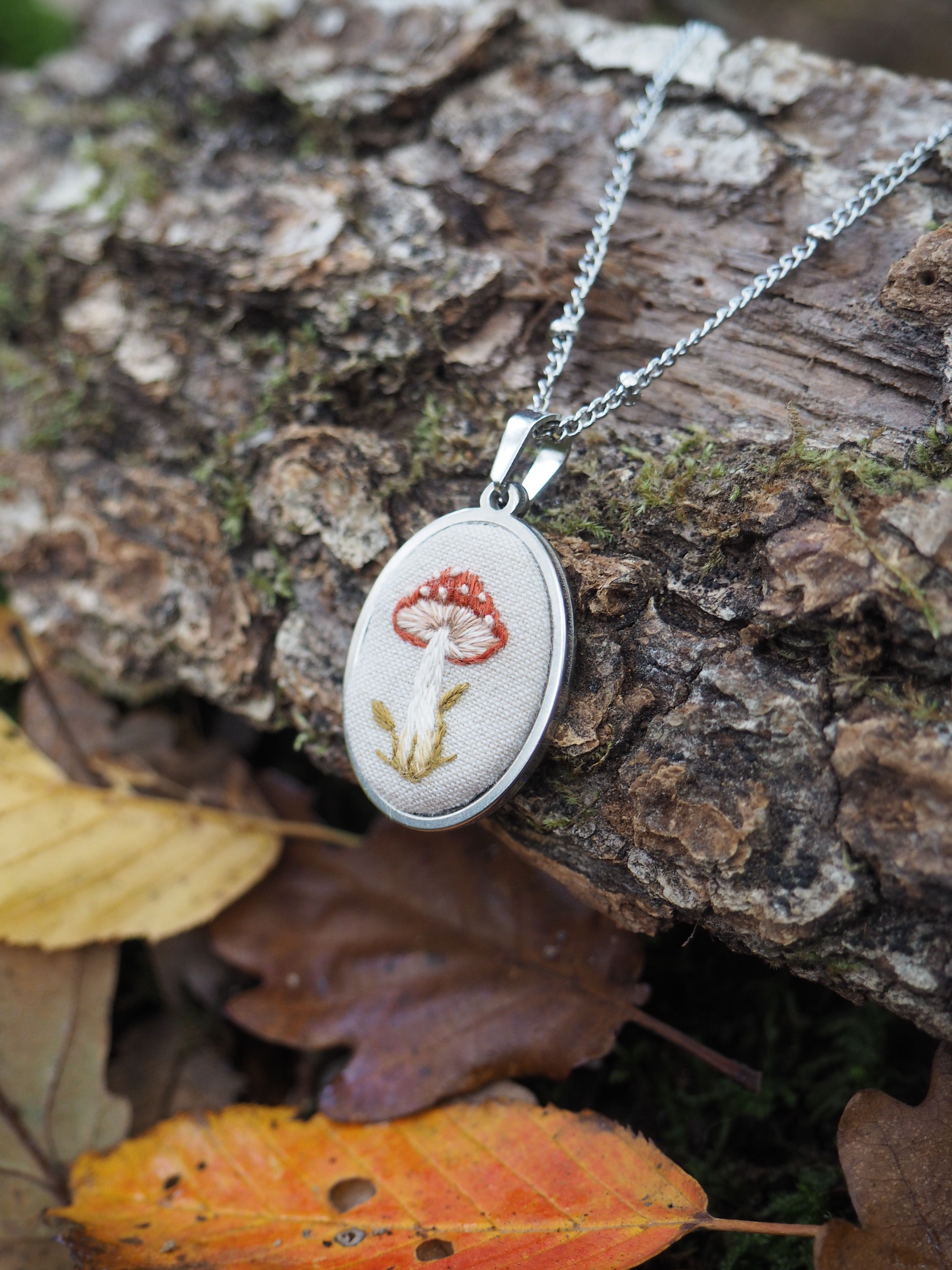 Cette image présente un pendentif délicat posé sur un tronc d'arbre dans un environnement naturel automnal. Le bijou consiste en un médaillon ovale en métal argenté, suspendu à une fine chaîne également argentée.  Le pendentif arbore un motif charmant : un champignon rouge à pois blancs (probablement une amanite tue-mouches) avec sa tige beige, brodé ou imprimé sur un fond blanc. Ce design évoque l'univers féerique et la nature sauvage. L'écorce rugueuse et texturée du tronc sur lequel repose le bijou crée un contraste intéressant avec la finesse du pendentif. Autour, on aperçoit quelques feuilles d'automne aux teintes chaudes - orange et jaune - qui parsèment le sol, ainsi qu'une touche de mousse verte, ajoutant à l'atmosphère naturelle et saisonnière de la composition. L'éclairage naturel met en valeur les détails du pendentif tout en créant une ambiance douce et organique, parfaitement en harmonie avec le thème champignon du bijou.