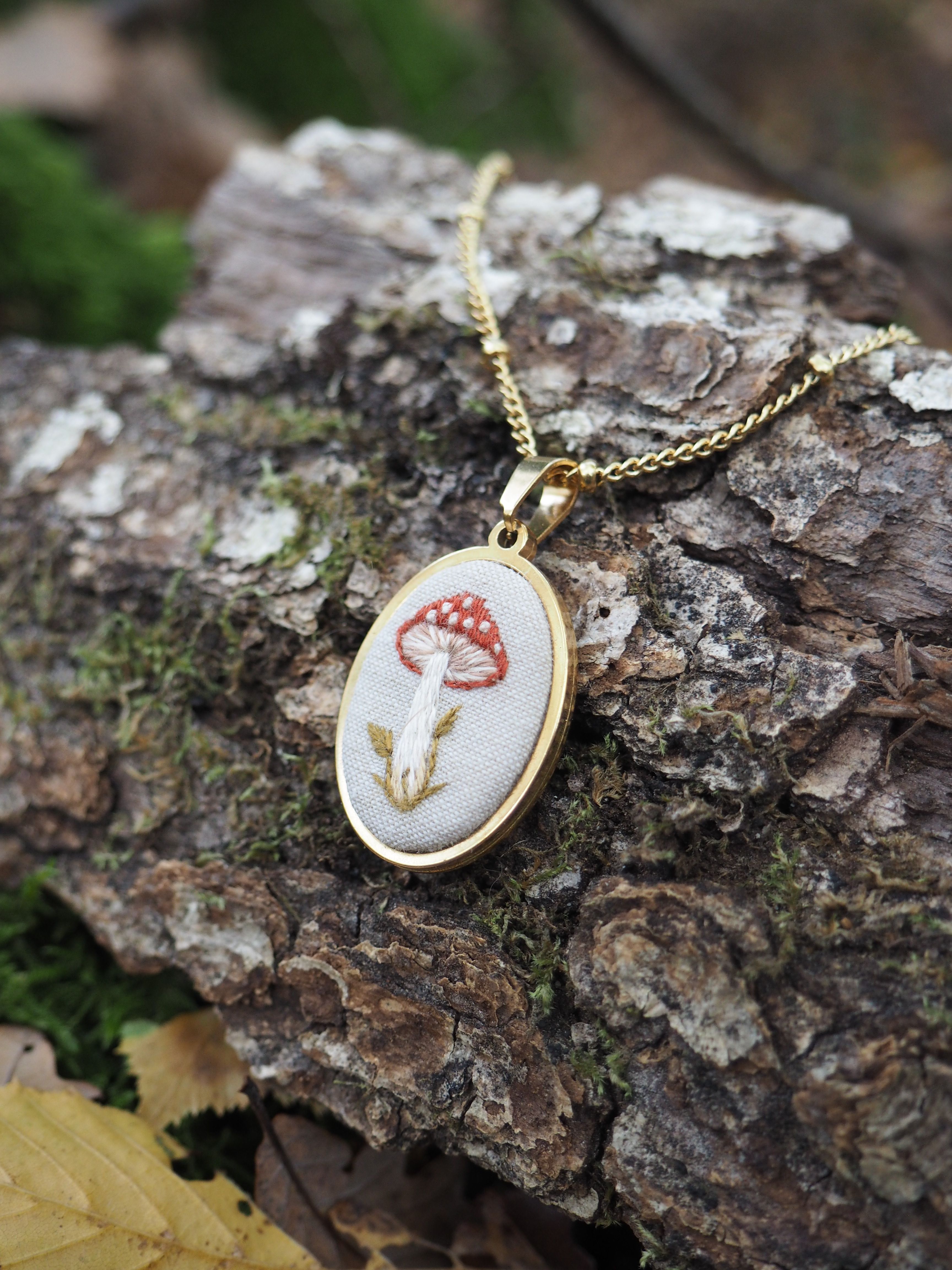 Cette image présente un délicat collier posé sur un morceau d'écorce d'arbre dans un environnement naturel. Le pendentif est de forme ovale avec un cadre doré élégant, et il contient une broderie ou un motif textile représentant un champignon rouge à pois blancs - très probablement une amanite tue-mouches avec sa couleur rouge caractéristique et ses taches blanches distinctives. Le champignon est accompagné de ce qui semble être une petite tige ou branche brodée en fil doré ou beige. L'ensemble du motif est réalisé sur un fond de tissu blanc ou crème, créant un contraste harmonieux avec les couleurs vives du champignon. Le collier repose sur une surface d'écorce rugueuse et texturée, avec de la mousse verte visible, ce qui crée une mise en scène très naturelle et poétique. L'arrière-plan est artistiquement flouté, mettant en valeur le bijou comme point focal de l'image. Ce type de bijou évoque un style artisanal, peut-être fait main, avec une esthétique qui rappelle les contes de fées et la nature sauvage.