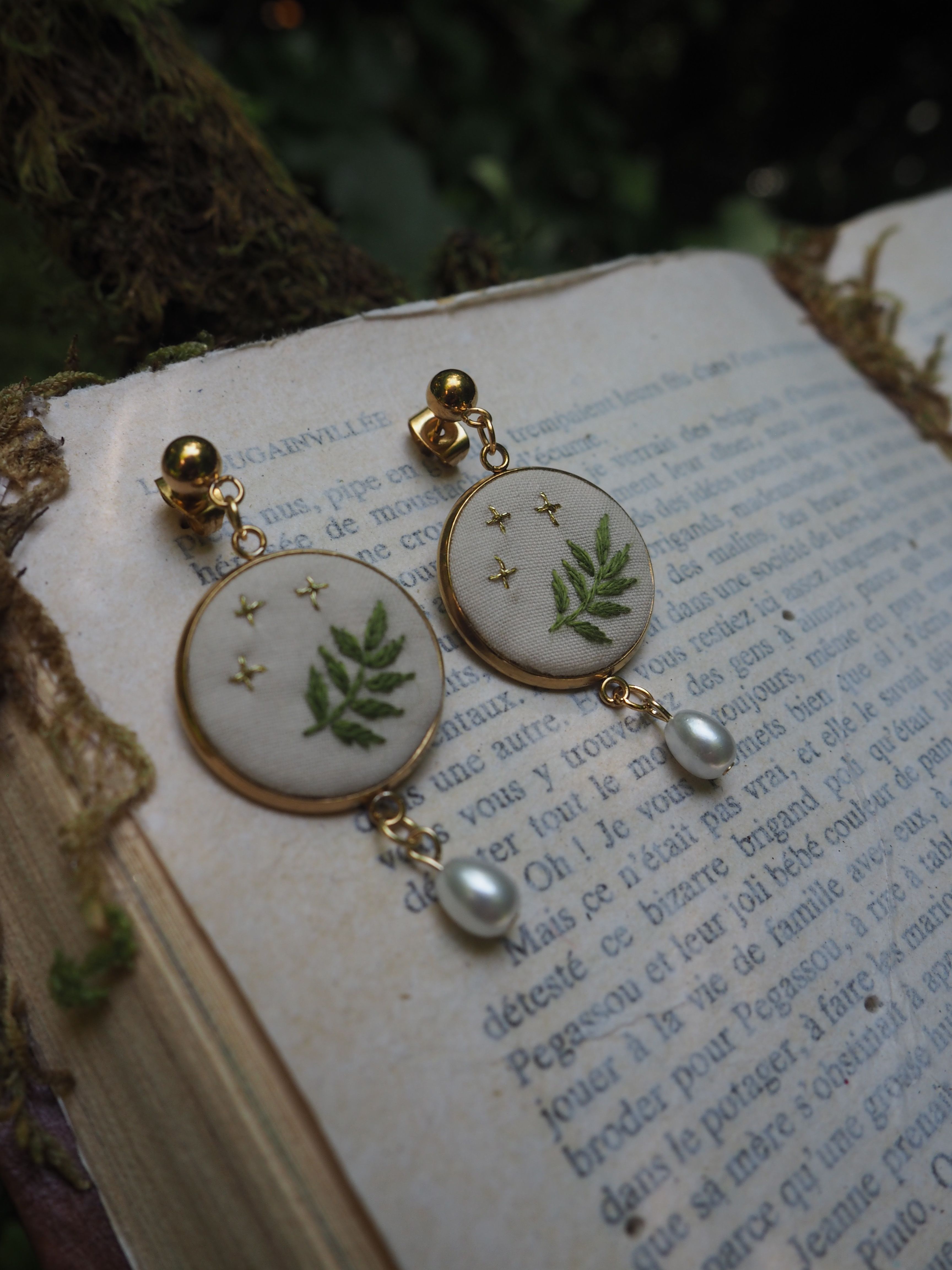 L'image montre une paire de boucles d'oreilles élégantes posées sur un livre ouvert. Les boucles d'oreilles sont composées de médaillons ronds blancs décorés de motifs de fougères vertes et de petites étoiles dorées. Au bas de chaque médaillon pend une petite perle nacrée. Les boucles d'oreilles semblent être montées avec des clips ou des tiges dorées. L'arrière-plan montre les pages d'un livre ancien avec du texte en français, donnant à l'image une ambiance vintage et romantique. La composition est artistique, avec un éclairage doux et naturel qui met en valeur les détails délicats des bijoux. Ces boucles d'oreilles ont un style botanique et délicat qui évoque la nature et le printemps, avec leur design incorporant des éléments naturels comme les fougères.