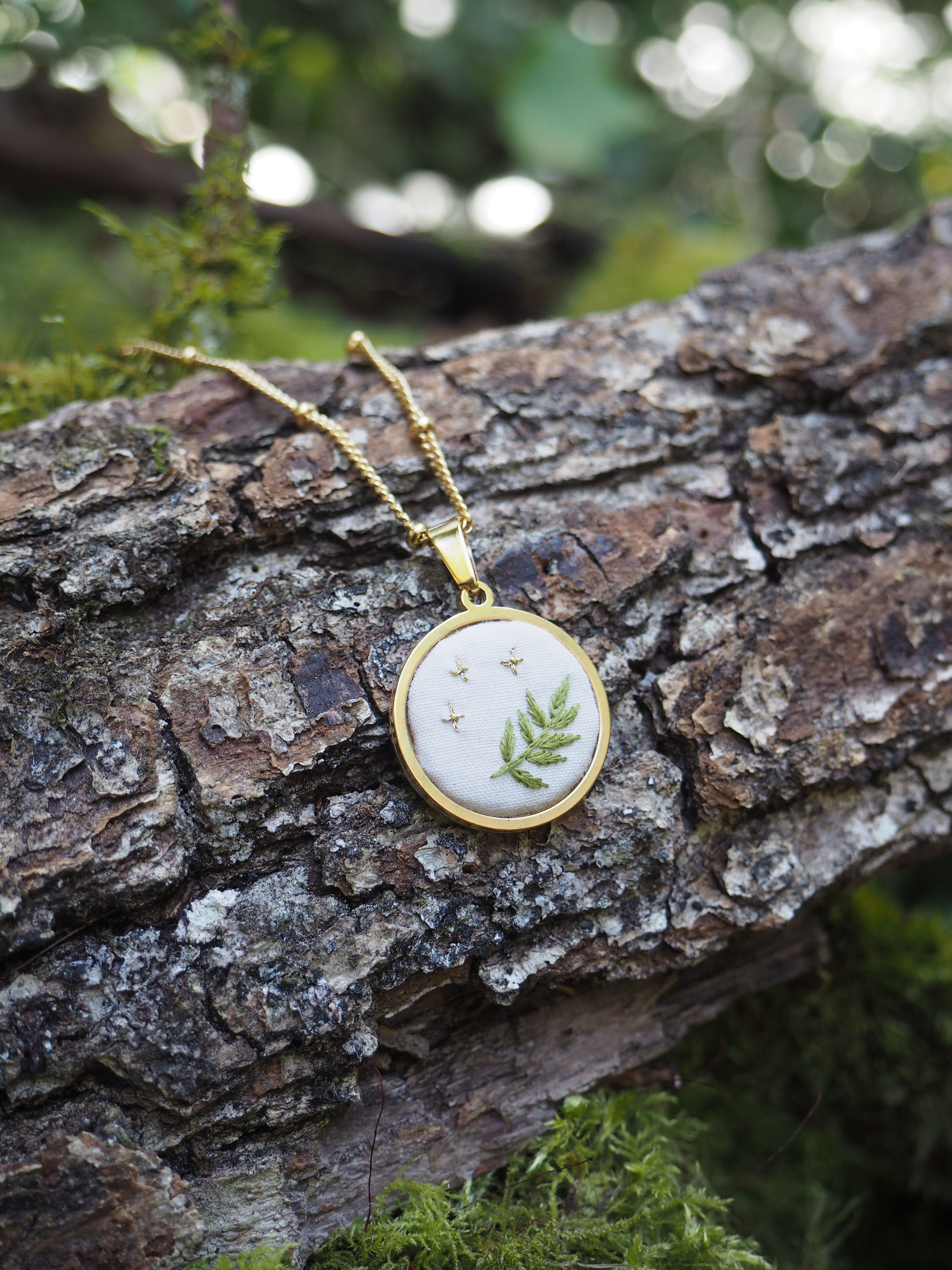 Cette image montre un délicat pendentif sur une chaîne dorée, posé sur un tronc d'arbre couvert d'écorce rugueuse. Le pendentif est rond, avec un cadre doré, et contient ce qui semble être une véritable fougère préservée sous verre ou résine. L'arrière-plan est flou mais on peut distinguer des tons verts naturels, suggérant un environnement forestier. La composition est très naturelle et artistique, créant un beau contraste entre le bijou raffiné et la texture brute de l'écorce. Ce type de bijou, qui incorpore des éléments botaniques réels, est souvent appelé "bijou botanique" ou "bijou terrarium".