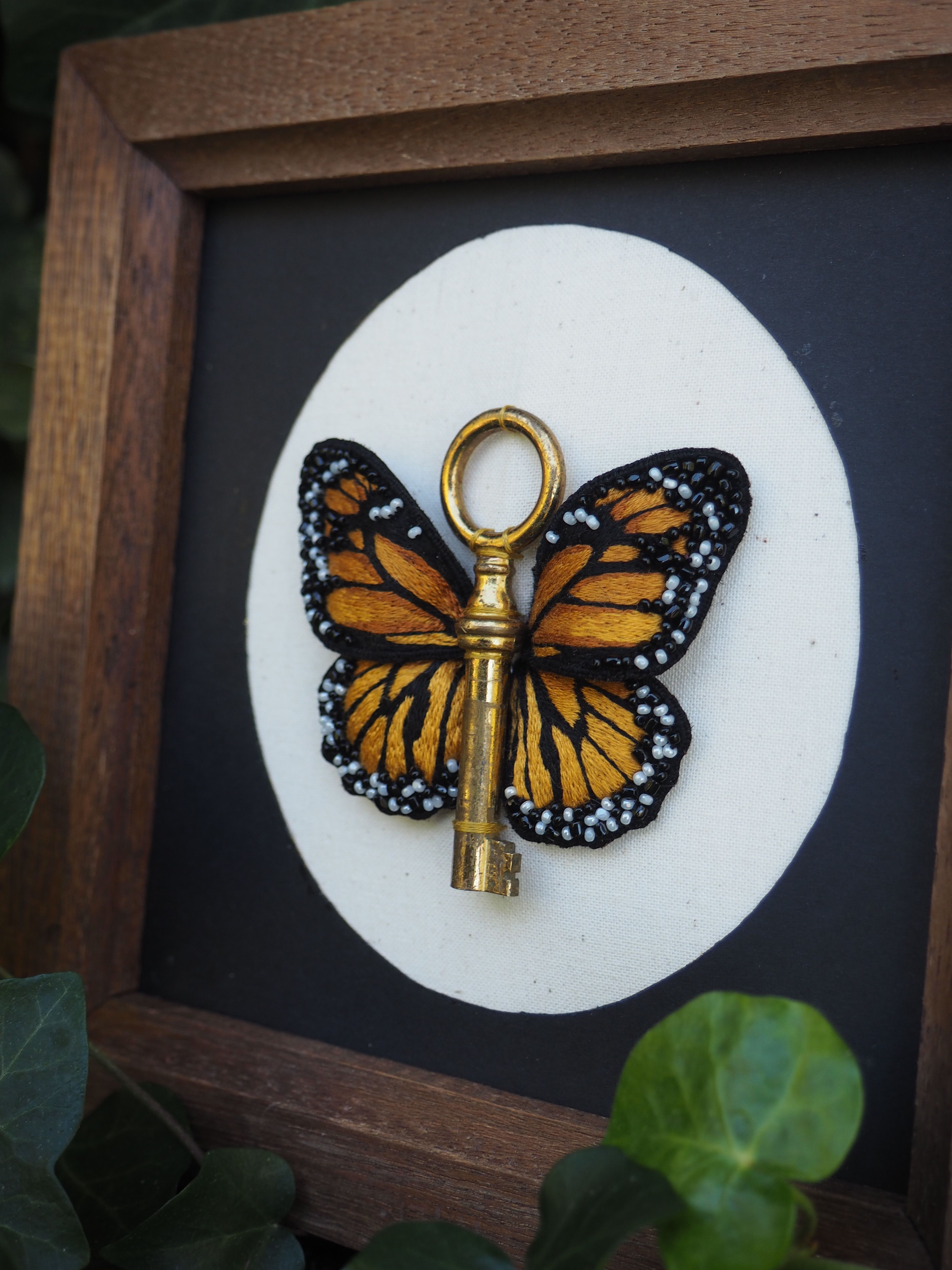 Cette image présente une composition artistique encadrée dans un cadre en bois foncé. Au centre, sur un fond noir, se trouve un cercle blanc qui met en valeur l'élément principal : une clé dorée ornée d'ailes de papillon monarque. La clé est de couleur dorée/laiton et possède un anneau circulaire à son extrémité. Les ailes de papillon, aux couleurs caractéristiques orange vif avec des bordures noires et des points blancs, sont fixées à la clé, créant un objet décoratif unique qui évoque la transformation et l'ouverture de nouvelles possibilités. Le contraste entre le fond noir et le cercle blanc fait ressortir cet objet symbolique. Dans les coins inférieurs du cadre, on aperçoit quelques feuilles vertes qui ajoutent une touche naturelle à cette présentation artistique. Cette pièce semble être une œuvre d'art conceptuelle qui joue sur les symboles de liberté (papillon), d'accès ou de découverte (clé), créant une métaphore visuelle poétique sur les thèmes de la transformation et des nouvelles opportunités.