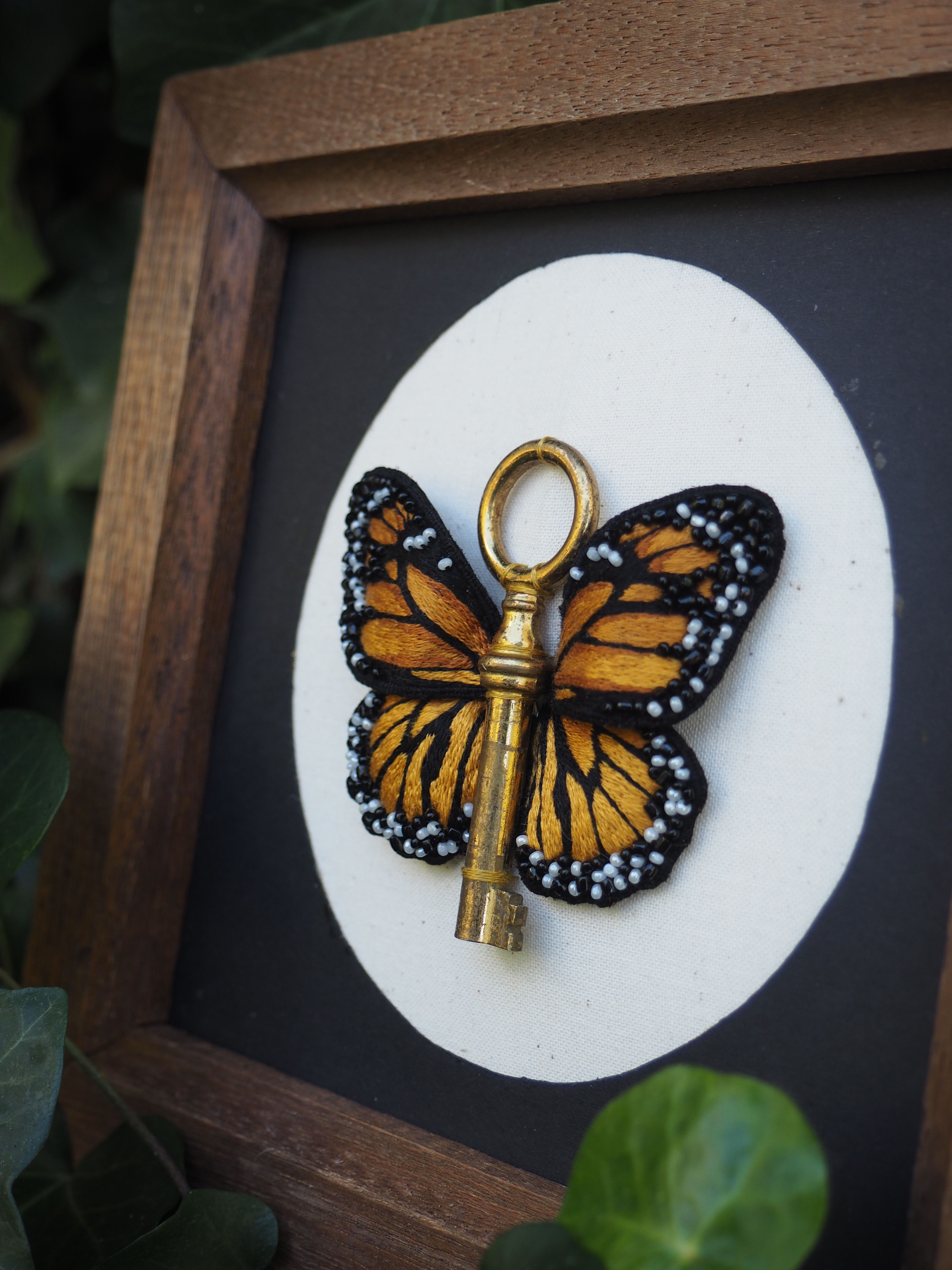 Cette image présente une œuvre d'art encadrée très créative et symbolique. Voici sa description détaillée : **Composition principale :**
- Un cadre en bois naturel de forme carrée
- Un fond noir mat qui fait ressortir l'élément central
- Un cercle blanc au centre servant de support à l'œuvre **Élément artistique central :**
- Une clé dorée/bronze transformée artistiquement
- Les ailes d'un papillon monarque (orange vif avec bordures noires et points blancs) ont été ajoutées à la clé
- La tête de la clé forme le "corps" du papillon
- L'anneau de la clé sert d'élément de suspension décoratif **Symbolisme :**
Cette création combine deux symboles puissants :
- La clé : représentant l'accès, les solutions, les opportunités
- Le papillon monarque : symbolisant la transformation, la liberté, la métamorphose **Contexte :**
L'œuvre est photographiée sur ce qui semble être une surface naturelle avec de la végétation visible en arrière-plan, suggérant un environnement extérieur ou près d'une fenêtre. Cette pièce artistique unique mélange habilement l'utilitaire et le naturel pour créer une métaphore visuelle sur la transformation et les nouvelles possibilités.