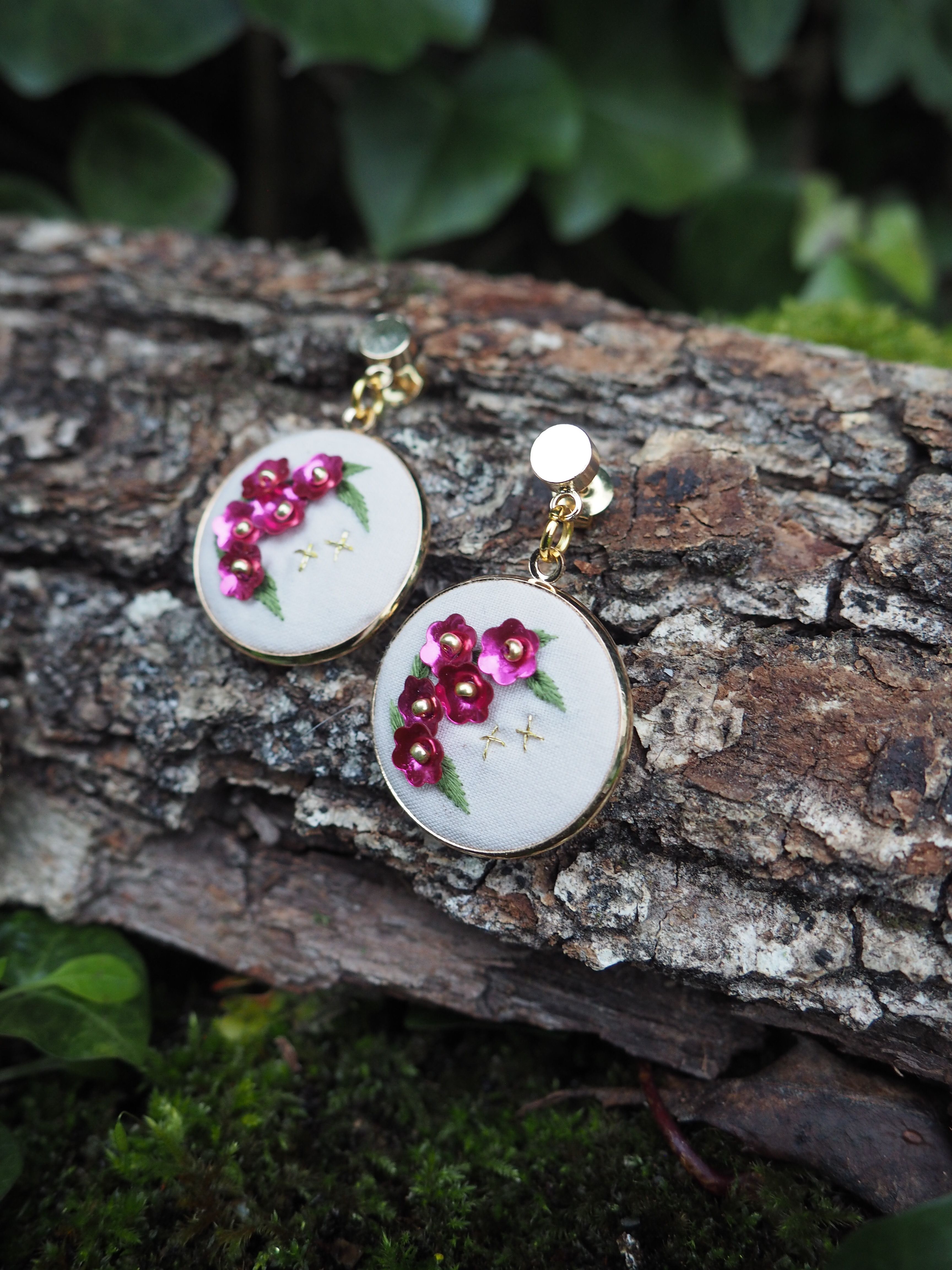 L'image montre une paire de boucles d'oreilles artisanales posées sur un morceau d'écorce d'arbre. Ces boucles d'oreilles sont de forme ronde et semblent être faites de tissu blanc brodé. Sur chaque boucle d'oreille, on peut voir de délicates fleurs brodées en rose foncé, probablement des roses ou des fleurs similaires. Les boucles d'oreilles ont une apparence très délicate et féminine. Elles sont attachées à des tiges dorées, ce qui suggère qu'elles sont conçues pour être portées aux oreilles. Le contraste entre la texture rugueuse de l'écorce et la finesse des broderies est frappant. L'arrière-plan montre un environnement naturel flou, avec des feuilles vertes visibles, ce qui donne à l'image une ambiance forestière ou de jardin. Cette mise en scène met en valeur la beauté naturelle et artisanale des bijoux. Cette photographie semble mettre l'accent sur la délicatesse du travail manuel et l'harmonie entre les créations artisanales et la nature.
