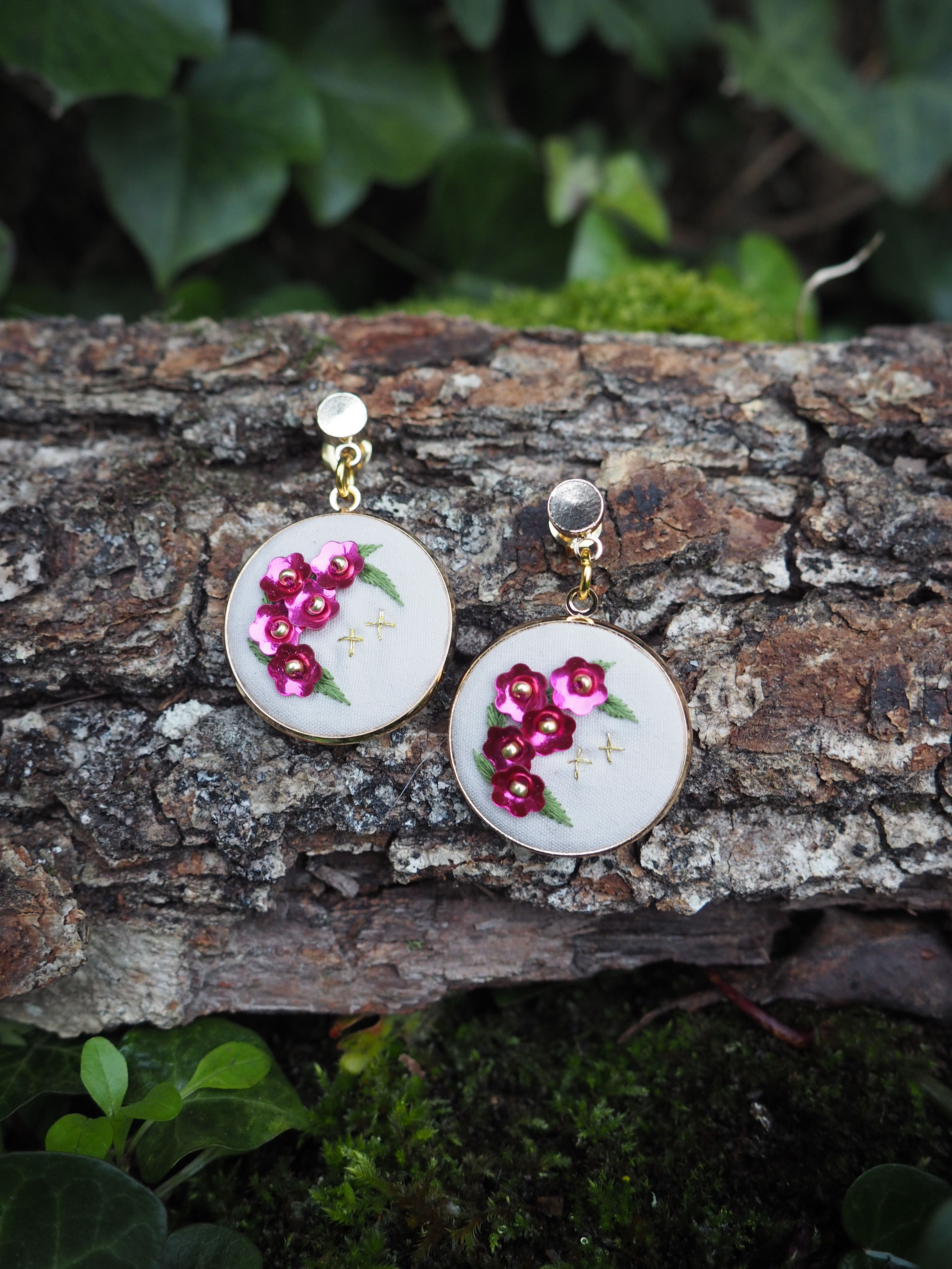 L'image montre une paire de boucles d'oreilles artisanales posées sur un morceau d'écorce d'arbre. Les boucles d'oreilles sont constituées de petits cercles blancs sur lesquels sont brodées des fleurs roses. Ces fleurs semblent être des cerisiers en fleurs ou des fleurs de pommier, brodées avec du fil rose vif. Les pendentifs sont attachés à des tiges dorées, probablement en métal. L'arrière-plan montre l'écorce rugueuse et texturée de l'arbre, avec quelques feuilles vertes floues visibles autour des bords de l'image, suggérant un cadre naturel en plein air. Cette photographie met en valeur le contraste entre les délicates boucles d'oreilles faites à la main et la texture brute de l'écorce, créant une composition intéressante qui mêle artisanat et nature. L'image semble capturer un moment de présentation créative de bijoux artisanaux dans un environnement naturel.