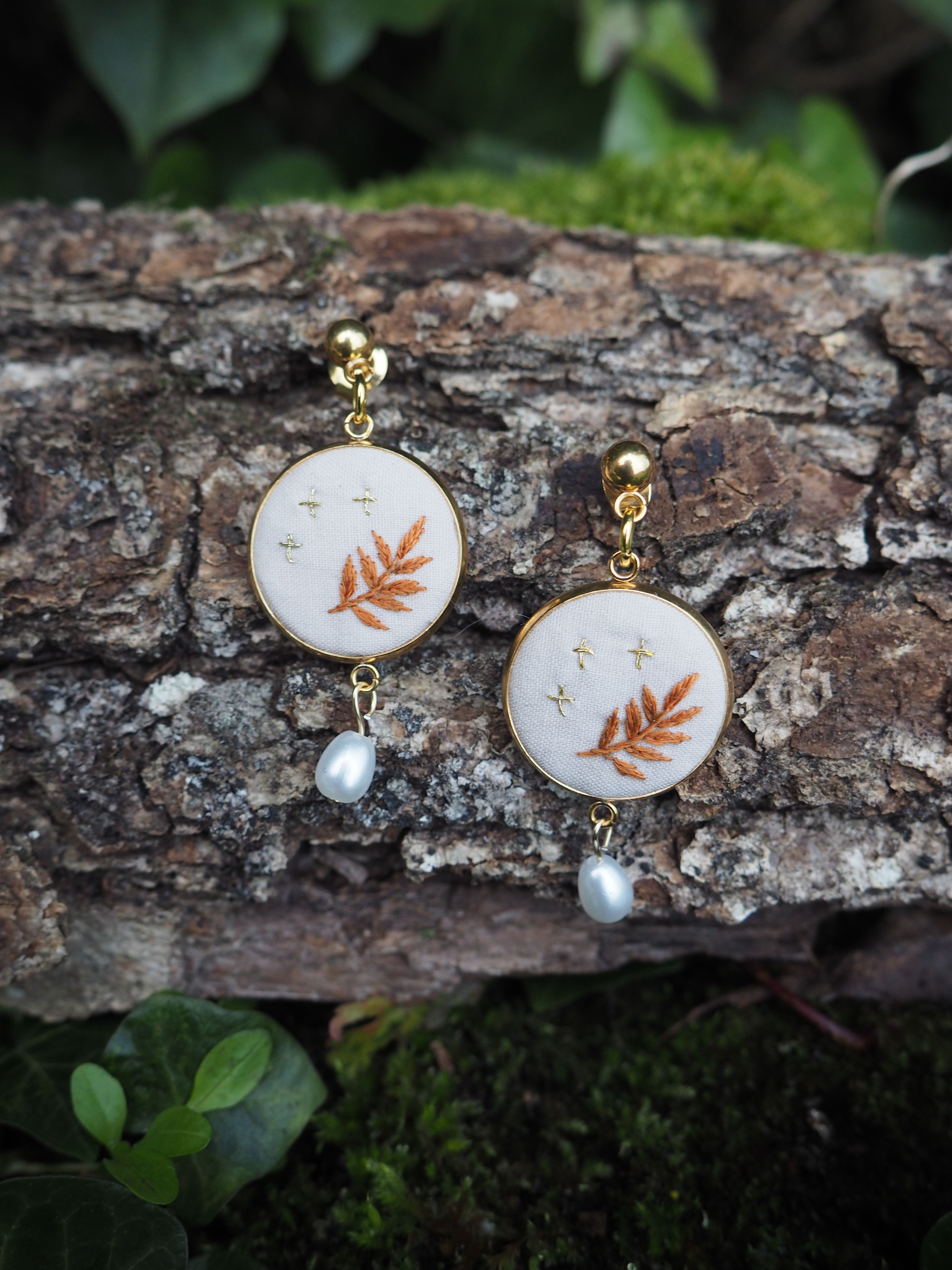 L'image montre une paire de boucles d'oreilles délicates posées sur un morceau d'écorce d'arbre. Les boucles d'oreilles sont composées de petits cercles blancs sur lesquels est brodé un motif de feuille orange ou cuivré. Sous chaque cercle pend une petite perle blanche. Les boucles d'oreilles semblent être faites à la main, avec un design naturel et organique qui s'harmonise bien avec le fond d'écorce rugueuse. L'arrière-plan montre également quelques feuilles vertes floues, suggérant un cadre naturel en extérieur. Le contraste entre la délicatesse des bijoux et la texture rugueuse de l'écorce crée une composition intéressante. Les couleurs douces des boucles d'oreilles - le blanc, l'orange pâle et l'éclat des perles - se détachent joliment sur les tons bruns de l'écorce. Cette image pourrait être utilisée pour présenter des bijoux artisanaux ou pour illustrer un style de mode inspiré par la nature.