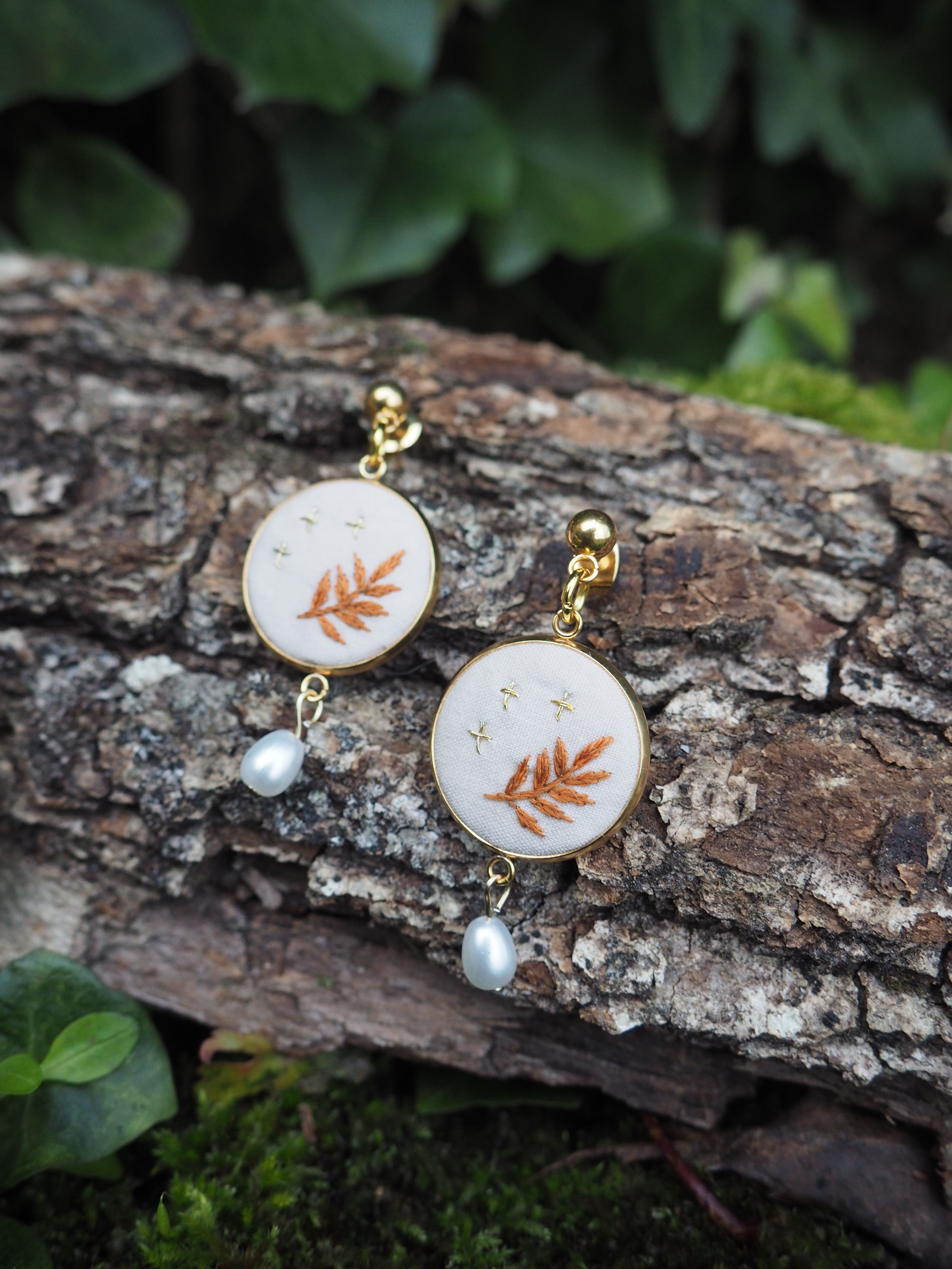 L'image montre une paire de boucles d'oreilles délicates posées sur un morceau de bois ou d'écorce. Les boucles d'oreilles sont composées de petits médaillons circulaires blancs, chacun orné d'une feuille de fougère de couleur cuivrée ou dorée. Sous chaque médaillon pend une petite perle blanche. Les boucles d'oreilles semblent être faites d'or ou d'un métal doré, ce qui contraste joliment avec le fond naturel rugueux de l'écorce. L'arrière-plan montre des feuilles vertes floues, suggérant que la photo a été prise dans un environnement naturel, peut-être un jardin ou une forêt. Cette image met en valeur la beauté délicate des bijoux inspirés par la nature, en les plaçant dans un cadre naturel qui complimente leur design. Le contraste entre les bijoux raffinés et la texture brute de l'écorce crée une composition intéressante et attrayante.