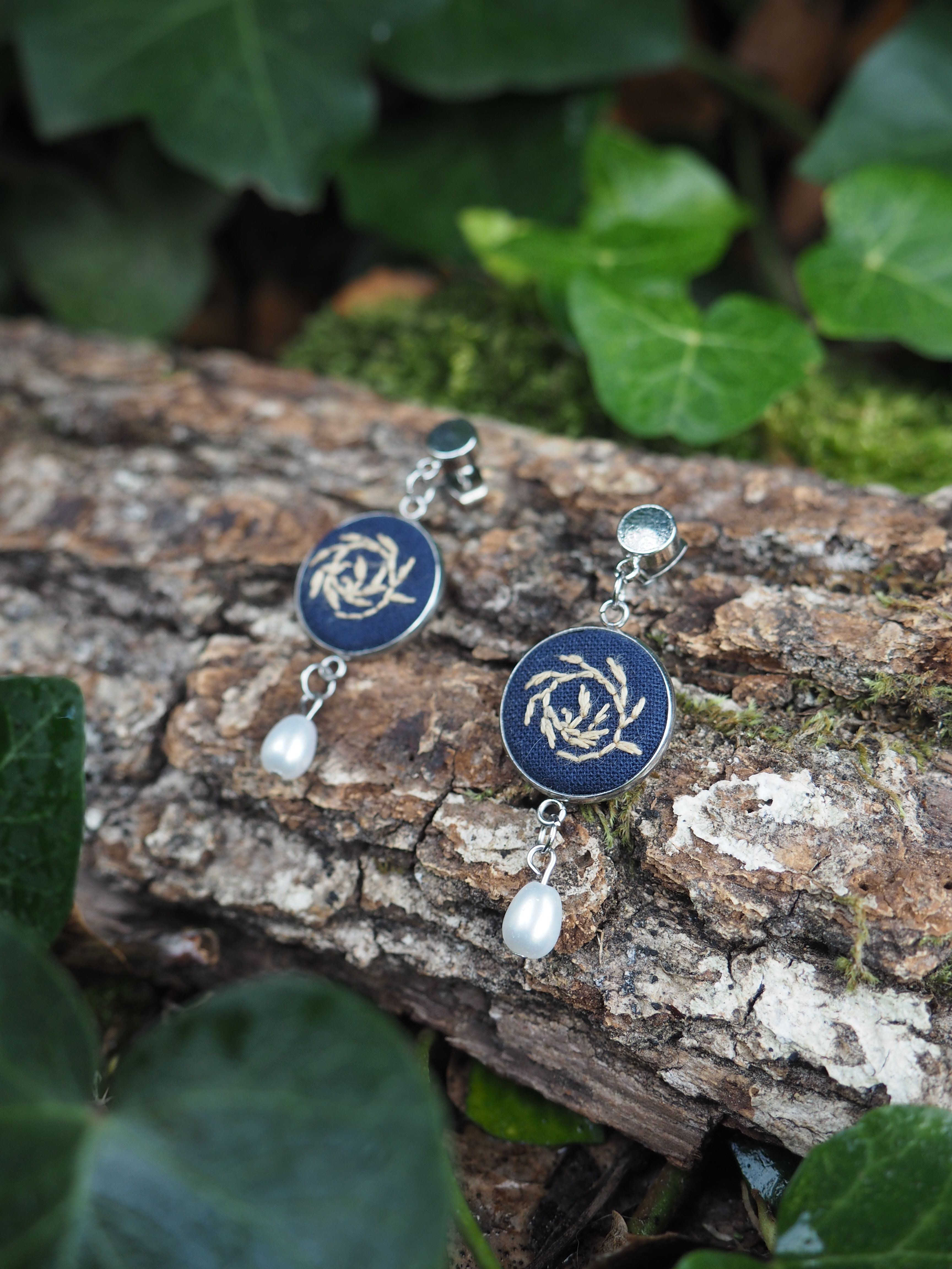 Cette image présente une paire de boucles d'oreilles élégantes disposées sur une branche d'arbre dans un environnement naturel.  **Description des bijoux :**
- Boucles d'oreilles pendantes avec des éléments circulaires bleu marine ornés de motifs décoratifs argentés en spirale ou entrelacs
- Perles blanches nacrées suspendues en pendentif
- Attaches et connecteurs en métal argenté
- Style artisanal avec une esthétique à la fois moderne et intemporelle **Mise en scène :**
- Les bijoux reposent sur une branche d'arbre rugueuse et texturée
- Arrière-plan composé de feuillage vert luxuriant légèrement flouté
- Éclairage naturel qui met en valeur les détails et les couleurs
- Composition photographique soignée créant un contraste harmonieux entre les éléments manufacturés et l'environnement naturel Cette présentation met particulièrement en valeur le caractère artisanal des bijoux tout en créant une atmosphère organique et apaisante.