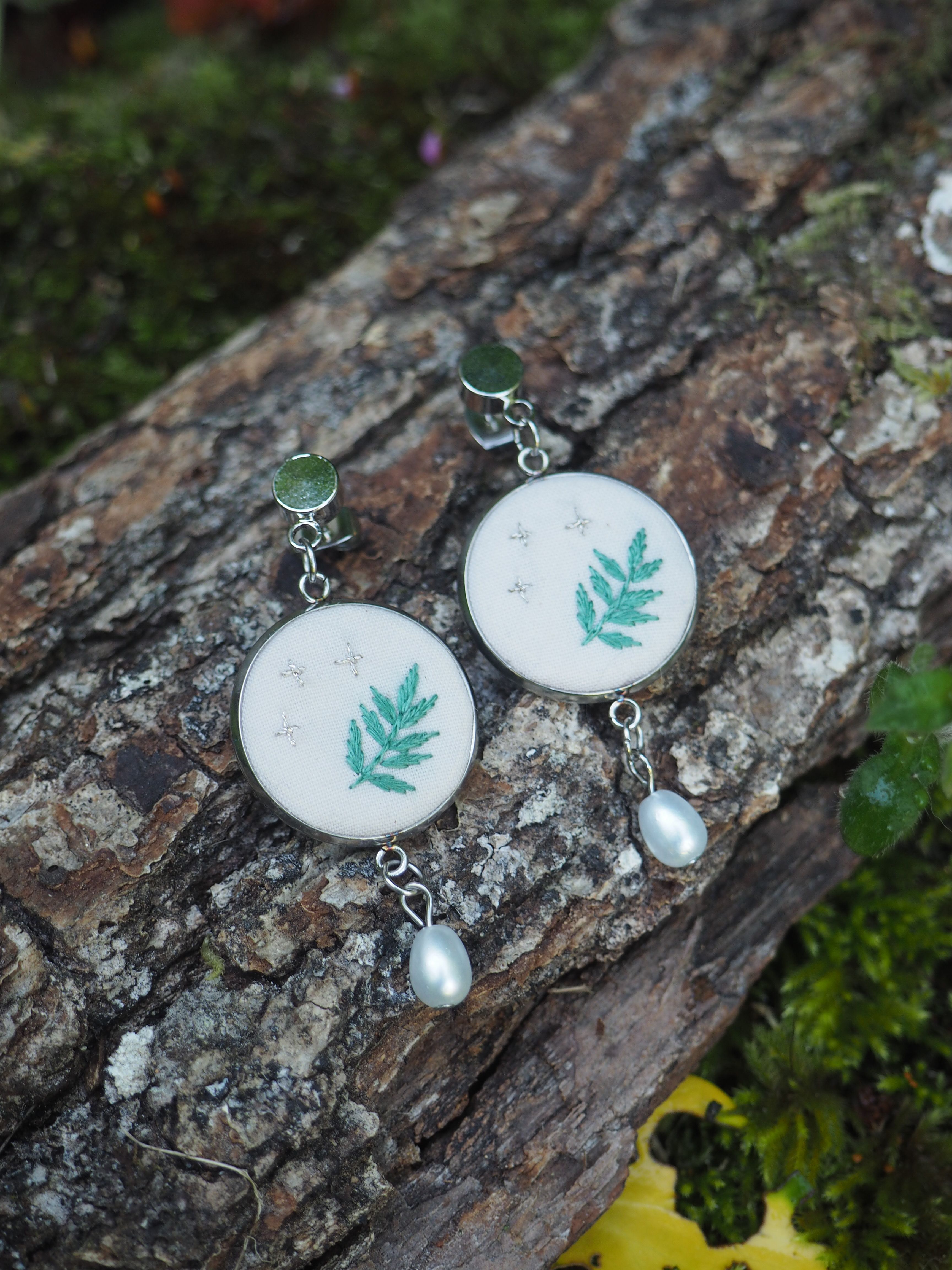 Ces boucles d'oreilles sont de délicates créations artisanales qui présentent un motif de feuille turquoise brodé ou imprimé sur un fond blanc circulaire. Chaque boucle d'oreille est ornée d'une perle nacrée suspendue au bas du médaillon principal. Les attaches semblent être en métal argenté avec de petites pierres ou cabochons verts. L'image montre les bijoux posés sur ce qui semble être un morceau de bois ou d'écorce texturé, avec de la mousse verte visible en arrière-plan, créant une ambiance naturelle et forestière qui complimente parfaitement le design botanique des boucles d'oreilles.