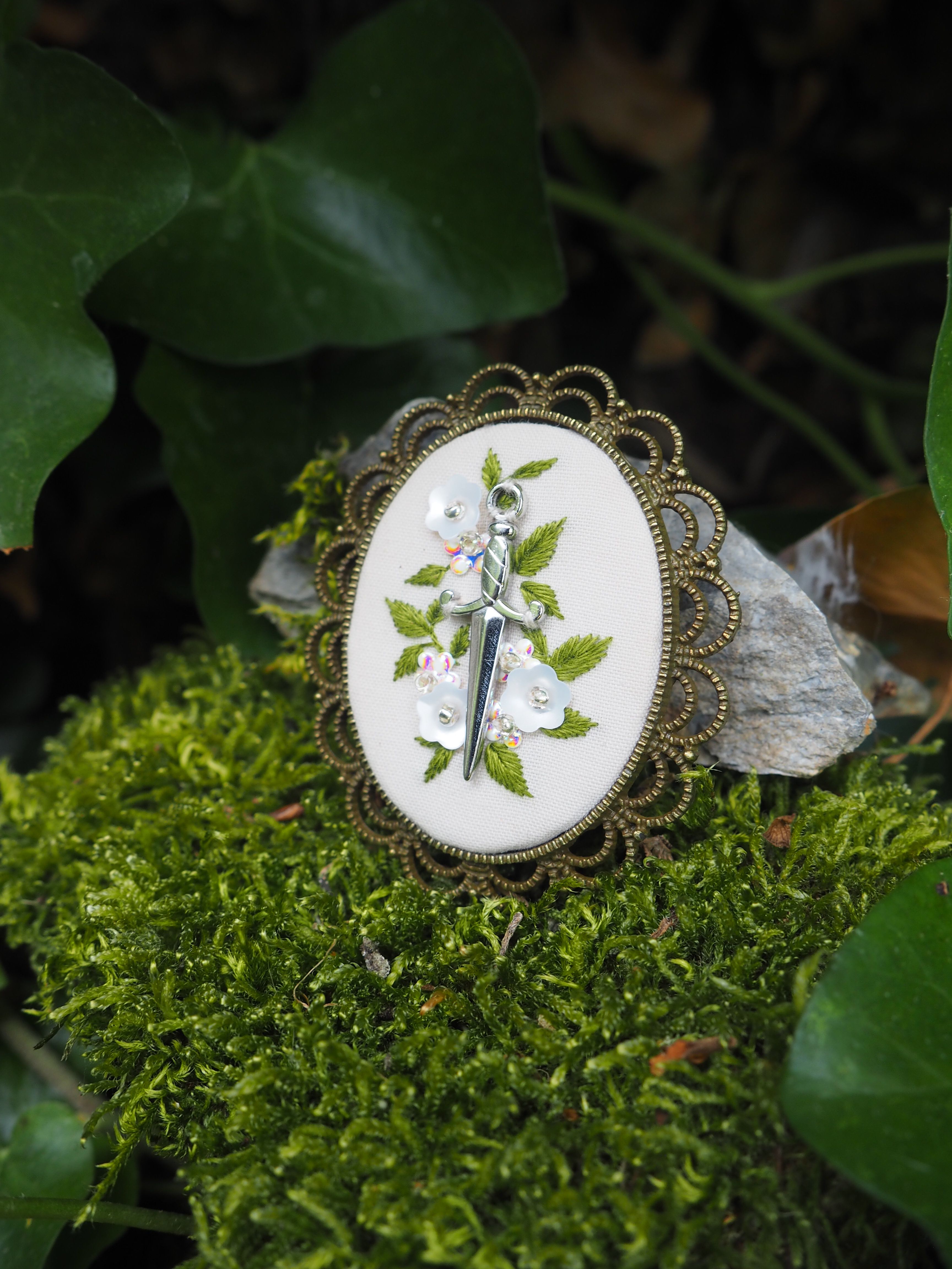 L'image montre des bijoux ou des accessoires décoratifs posés sur de la mousse verte. Ce sont des broches ou des pendentifs qui semblent être de style vintage ou antique, avec des bordures en métal doré finement ouvragées dans un style filigrane. Le centre de chaque pièce présente ce qui ressemble à des motifs botaniques ou des feuilles vertes sur un fond blanc. Les bijoux sont photographiés dans un cadre naturel, avec de la mousse verte fraîche et quelques feuilles en arrière-plan, créant une ambiance naturelle et romantique.