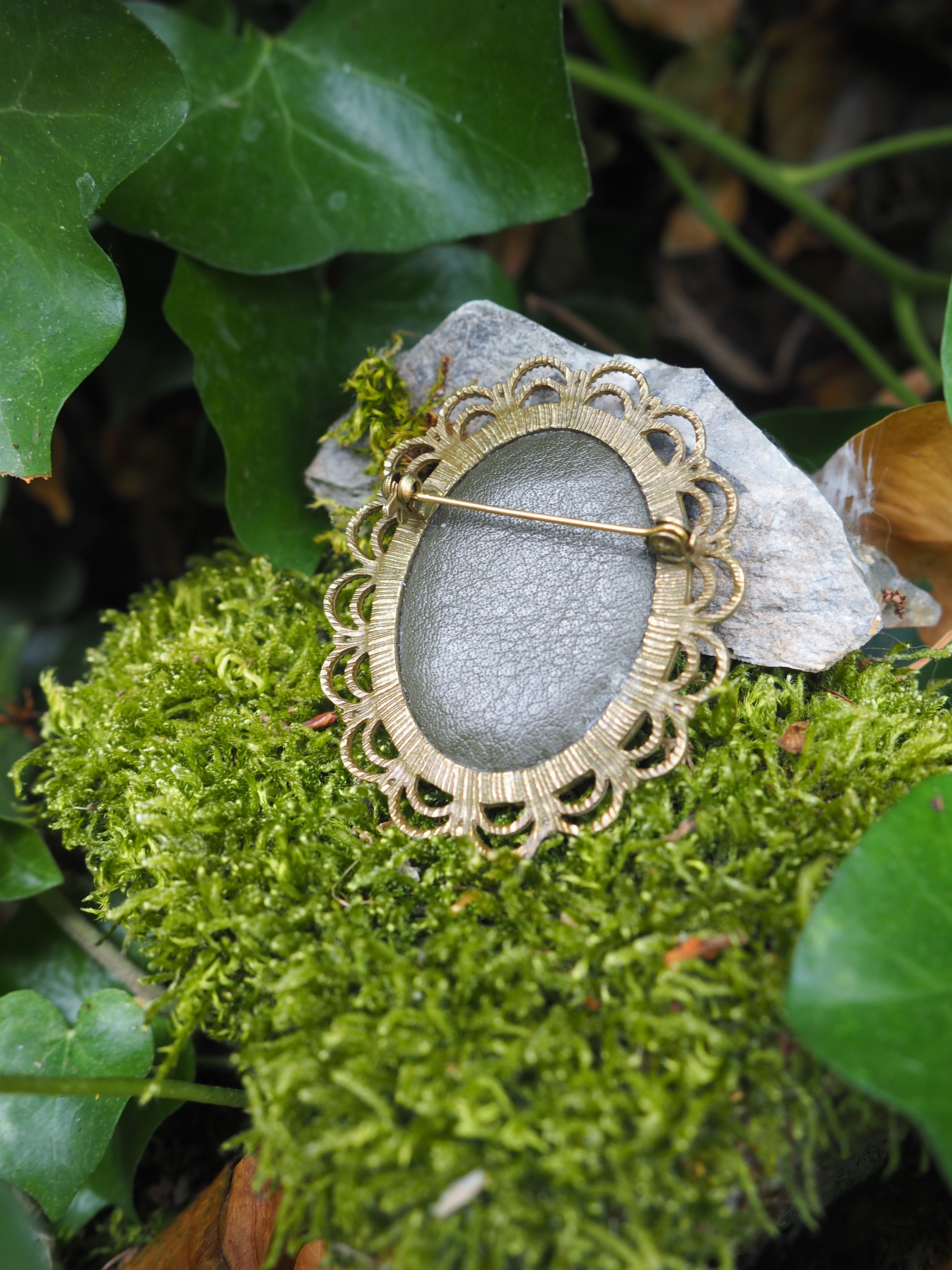 L'image montre des broches ou des bijoux vintage avec des motifs floraux décoratifs en métal doré, disposés sur un lit de mousse verte. Les broches semblent avoir des centres argentés ou gris mat, et leur design rappelle des fleurs stylisées avec des bordures ornementales. Le fond présente également quelques feuilles vertes, créant un cadre naturel et organique pour ces pièces de bijouterie délicates. L'ambiance de la photo est très romantique et évoque un style vintage ou antique.