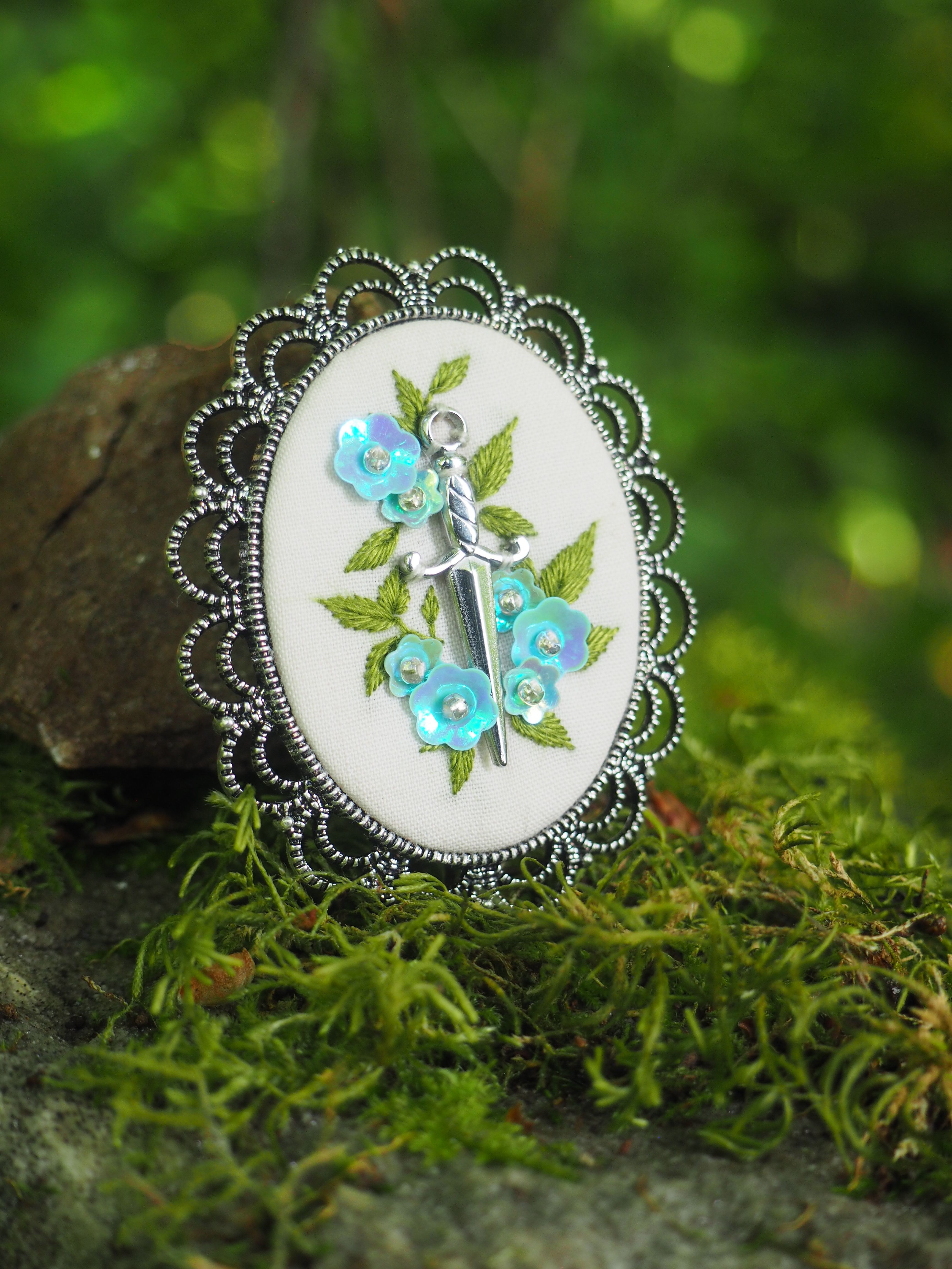 Cette image montre une belle broche ou un pendentif décoratif avec un cadre en métal argenté au motif festonné. Le centre présente une broderie délicate sur un fond blanc, représentant des fleurs bleues avec des feuilles vertes. Le design est élégant et délicat, avec un style vintage ou romantique. La photo est prise sur un fond naturel de mousse verte, ce qui crée un beau contraste avec le bijou et lui donne une ambiance féerique ou enchantée. L'éclairage doux et le cadre naturel mettent parfaitement en valeur les détails délicats de cette pièce artisanale.