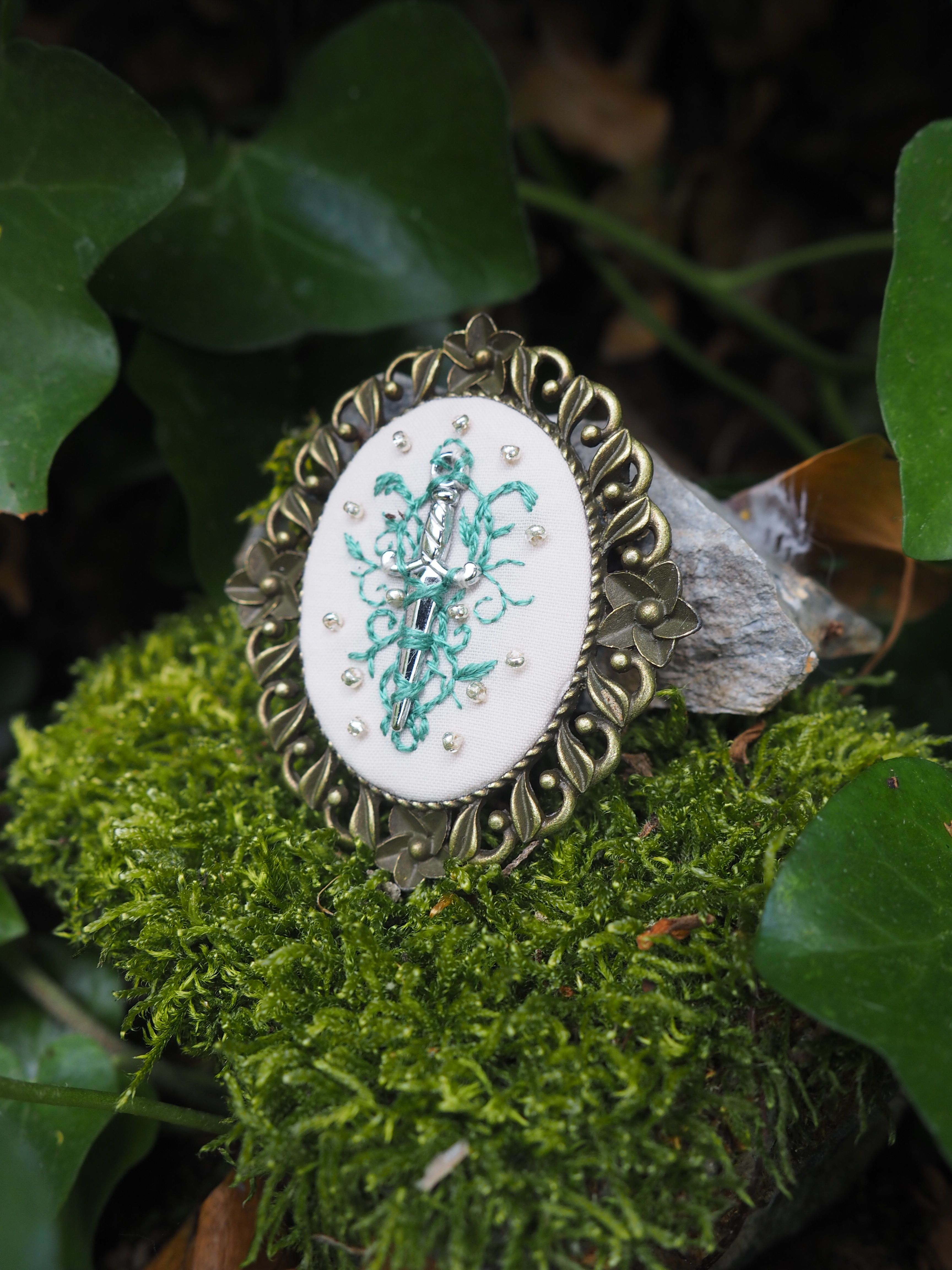 Cette image montre un joli bijou ou broche vintage posé sur de la mousse verte dans un cadre naturel. Le bijou présente un médaillon ovale avec un motif floral ou décoratif de couleur turquoise sur fond blanc, entouré d'un cadre ornementé en métal de style antique avec des motifs décoratifs. L'arrière-plan montre quelques feuilles vertes et de la mousse, donnant à la photo une ambiance naturelle et féerique. La composition est très artistique, mêlant l'aspect délicat du bijou avec les éléments naturels qui l'entourent.