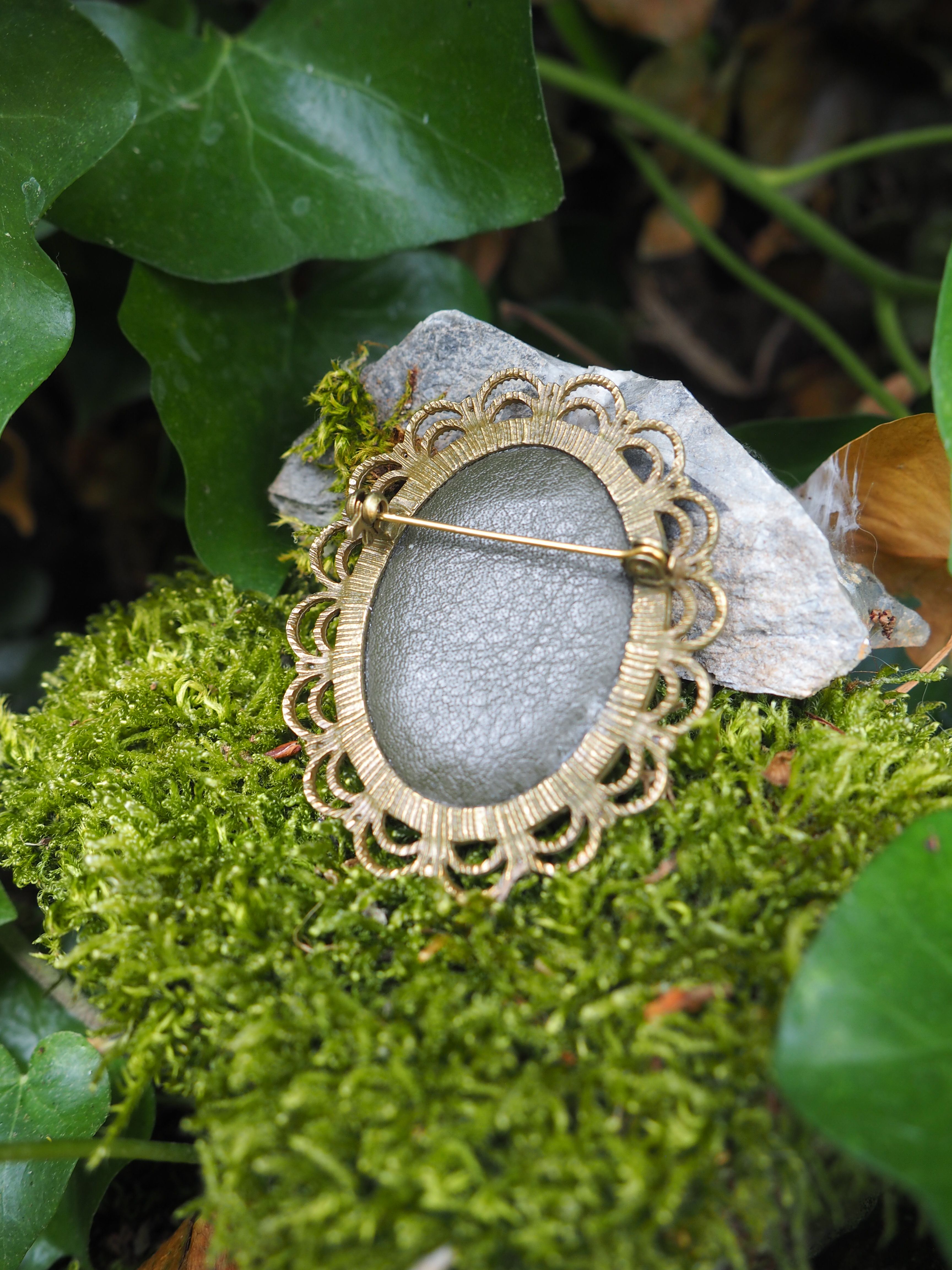 L'image montre des bijoux ou des broches vintage posés sur un lit de mousse verte. Les pièces semblent être des broches ou des pendentifs de style ancien avec des motifs décoratifs ornementaux autour de ce qui paraît être des pierres ou des cabochons gris/argentés au centre. Le cadre naturel avec la mousse verte et quelques feuilles en arrière-plan crée une ambiance féerique et romantique. Le contraste entre les éléments métalliques dorés des bijoux et la texture naturelle de la mousse verte est particulièrement esthétique.