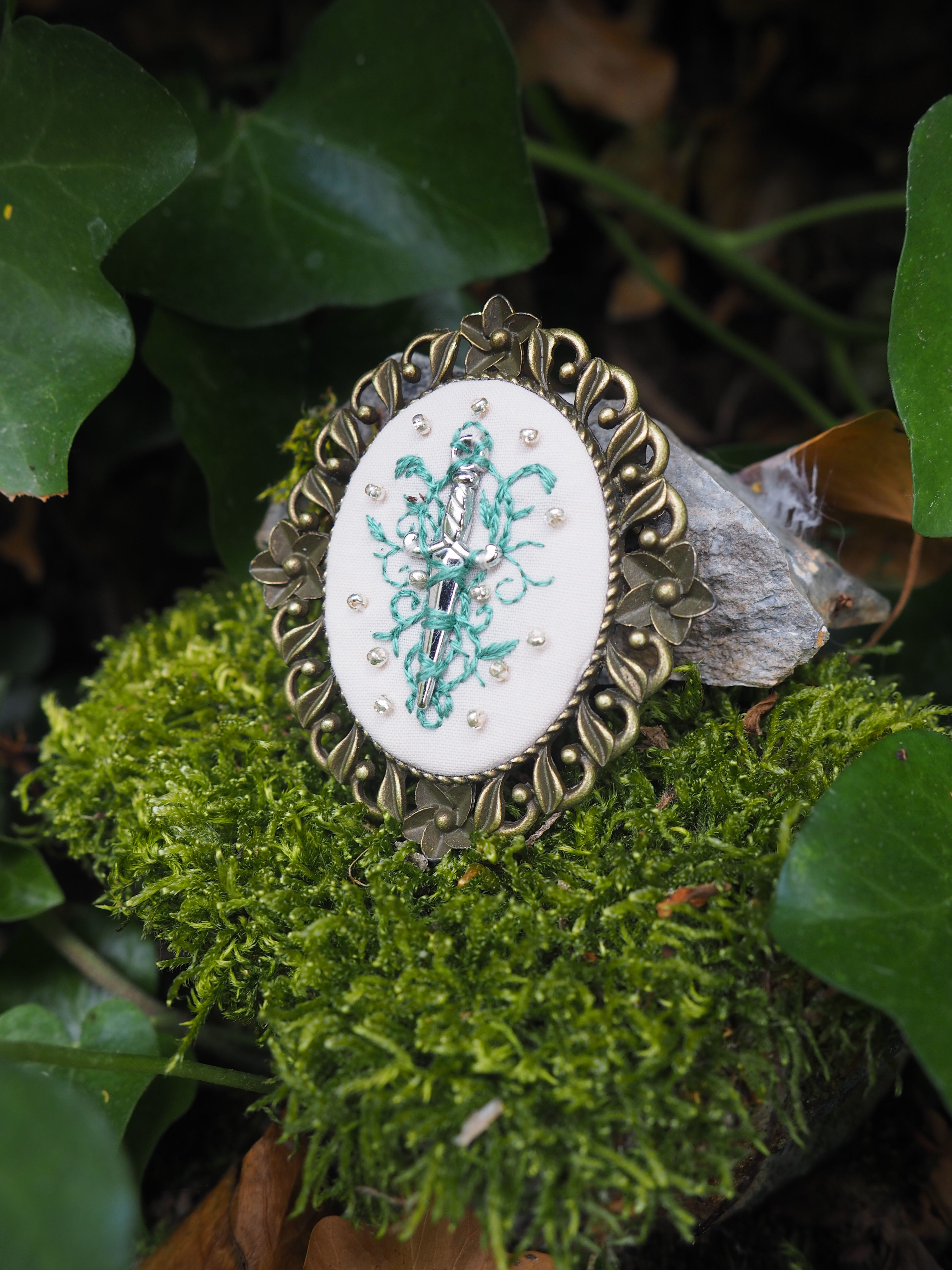 Cette image montre un joli bijou ou broche vintage posé sur un lit de mousse verte. Le bijou a un cadre décoratif en métal de style baroque ou victorien avec des motifs ornementaux. Au centre se trouve un médaillon rond avec une délicate broderie ou un dessin floral en turquoise sur fond blanc. L'ensemble est photographié dans un cadre naturel avec des feuilles vertes en arrière-plan, créant un contraste intéressant entre l'objet artisanal et l'environnement naturel. La composition est artistique et évoque une ambiance féerique ou romantique.