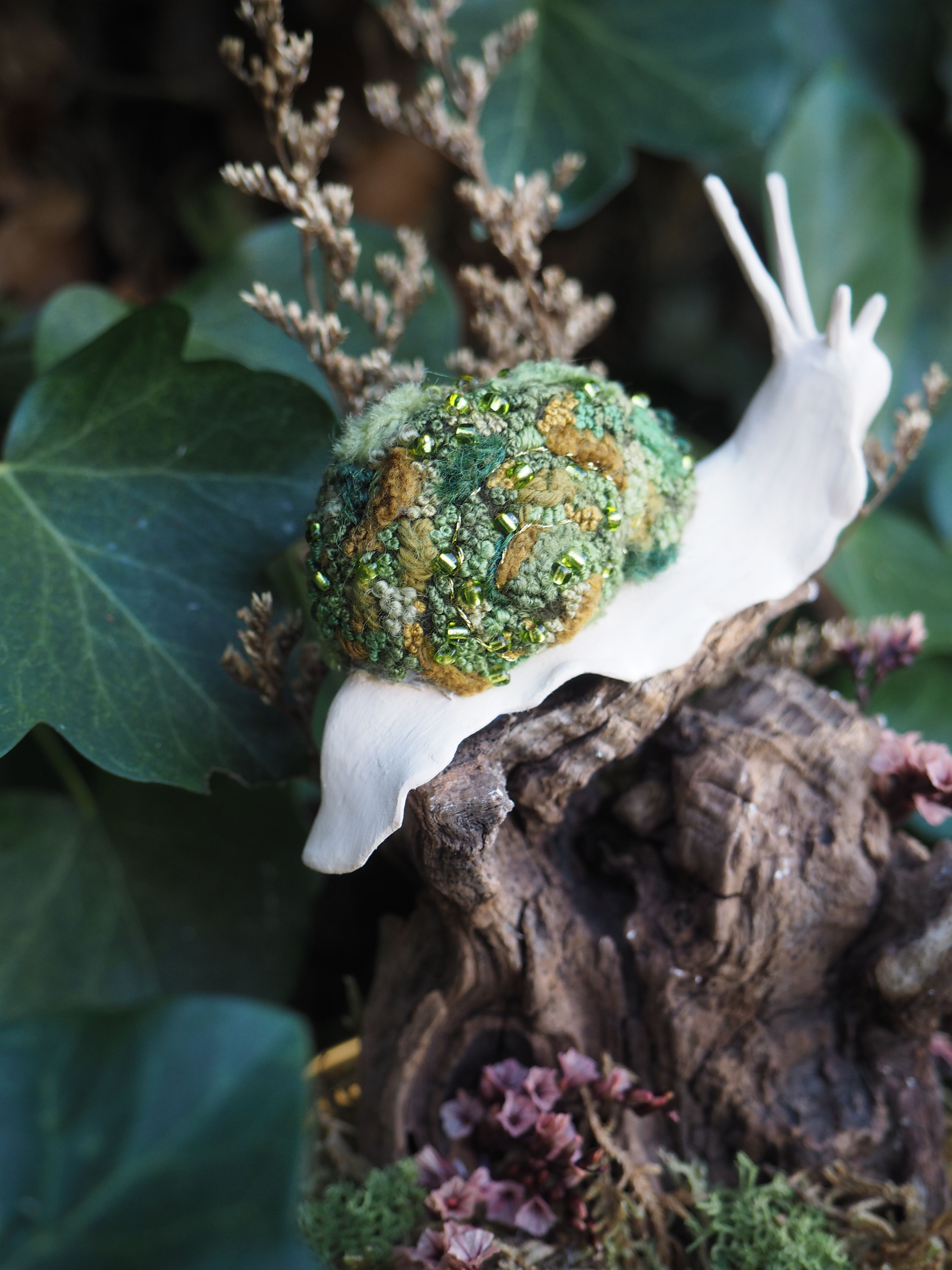 Cette image présente une broche en forme d'escargot, posée sur un morceau de bois flotté ou de souche, dans un environnement qui évoque la nature. L'escargot est l'élément central de l'image. Son corps est d'un blanc immaculé, lisse et mat, contrastant avec sa coquille. La coquille est particulièrement détaillée et texturée, composée de matériaux qui ressemblent à du feutre ou de la laine cardée, dans des nuances de vert et de brun. On y distingue des touches de vert clair, de vert forêt et de brun terreux, avec des petites perles ou des paillettes dorées et vertes incrustées, ajoutant un aspect scintillant et organique, comme de la mousse ou du lichen. Les antennes de l'escargot sont fines et blanches, prolongeant l'élégance de son corps. L'escargot est posé sur un morceau de bois sombre et texturé, qui semble être une souche ou une branche morte, avec des creux et des reliefs. Autour de la base du bois, on aperçoit des éléments végétaux, comme de la mousse verte et des petites fleurs séchées ou des lichens de couleur rosée ou violacée, ajoutant de la profondeur et de la couleur à la scène. En arrière-plan, on distingue un feuillage vert foncé, probablement du lierre, qui crée un fond flou et naturel, mettant en valeur l'escargot. Des éléments végétaux séchés de couleur beige ou marron clair sont également visibles, contribuant à l'ambiance forestière et automnale. La lumière semble douce et naturelle, éclairant l'escargot et ses détails, tout en laissant l'arrière-plan légèrement dans l'ombre, ce qui accentue la mise au point sur l'objet principal. L'ensemble de l'image dégage une atmosphère délicate et artistique, mettant en avant le travail artisanal de la broche.