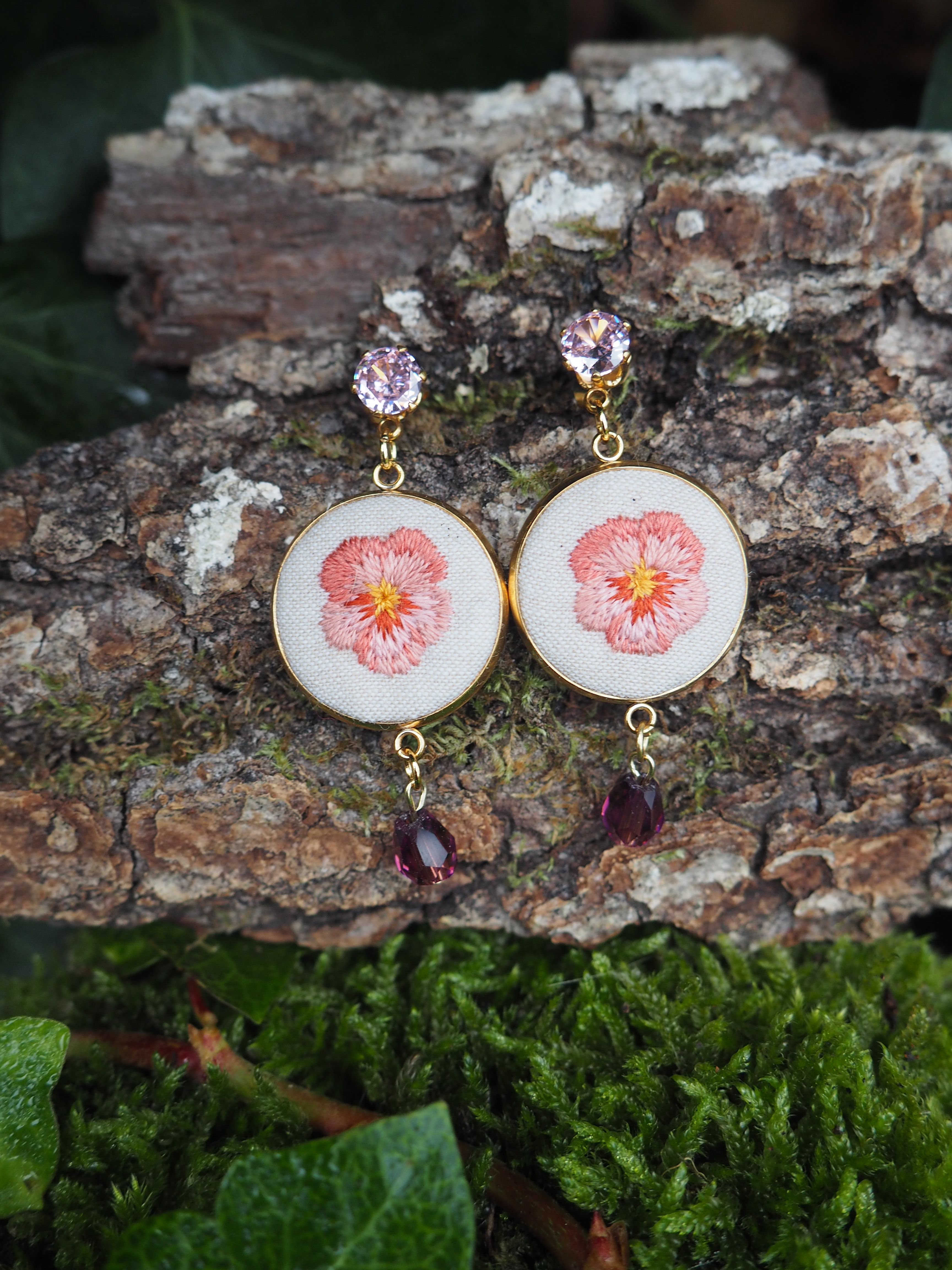 This close-up shot features a pair of handcrafted earrings, each showcasing a delicate embroidered flower, likely a pansy, in shades of pink and orange on a white fabric background. The embroidery is encased in a gold-toned circular frame. Above the embroidered circle, each earring is attached to a small, faceted, light purple or lavender gemstone, which serves as the stud or top part of the earring. Below the embroidered circle, a darker, faceted, teardrop-shaped gemstone, possibly amethyst or a similar purple stone, dangles from a small gold-toned loop. The earrings are displayed against a natural backdrop. They rest on a piece of textured tree bark, which has patches of light green moss growing on it. In the foreground and background, there are hints of dark green foliage, possibly ivy, adding to the organic and rustic aesthetic of the presentation. The lighting appears soft and natural, highlighting the details of the embroidery and the sparkle of the gemstones.