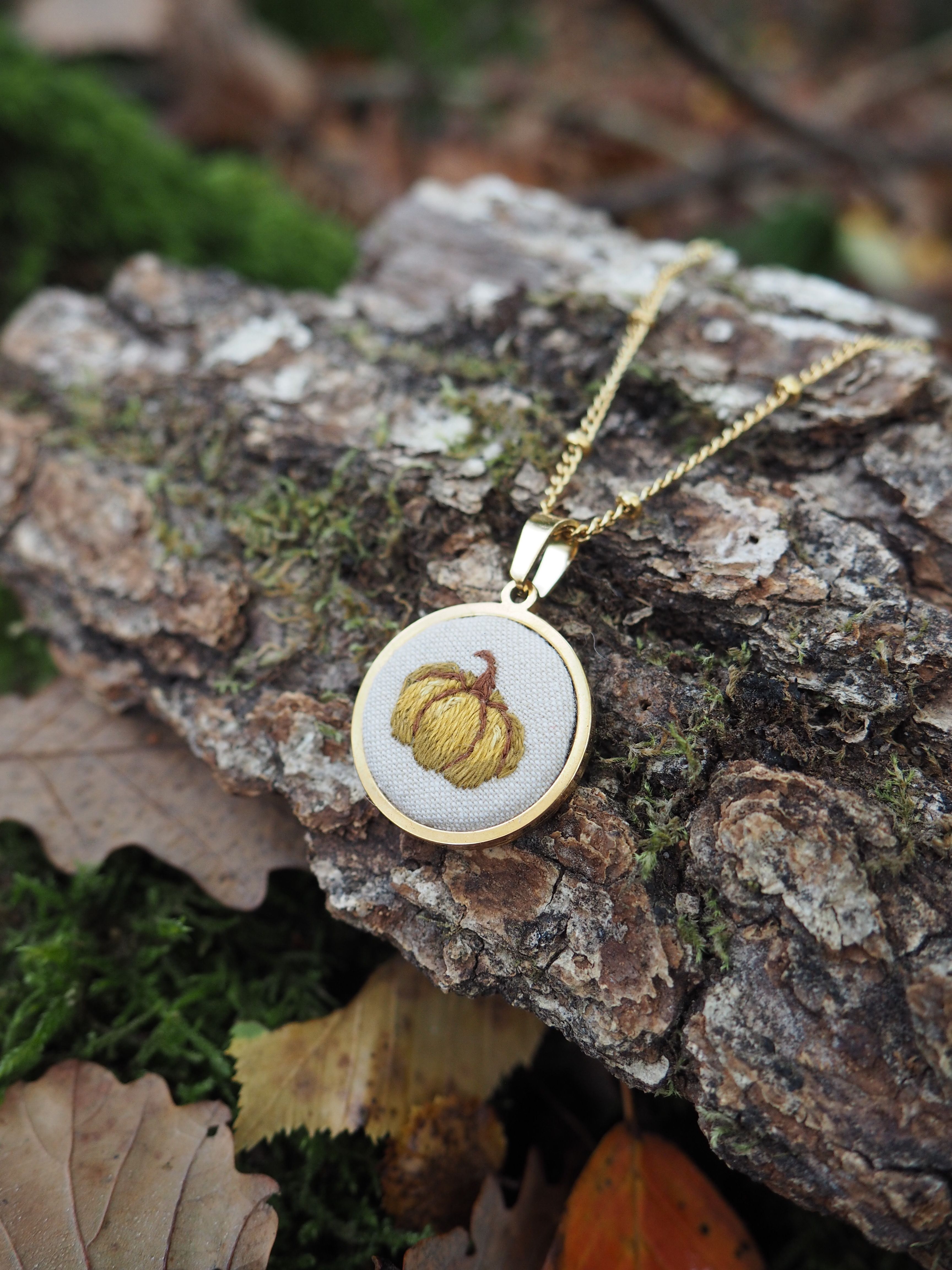 Cette image présente un bijou élégant posé sur un environnement naturel. Il s'agit d'un pendentif circulaire suspendu à une chaîne dorée fine. Le pendentif lui-même arbore un design bicolore avec un cadre doré entourant un centre de couleur claire (probablement blanc ou crème). Au centre du pendentif, on peut observer un motif décoratif qui ressemble à une citrouille ou un potiron stylisé, avec des détails dorés qui évoquent les nervures caractéristiques de ce légume. Le bijou repose sur un morceau d'écorce ou de bois naturel, créant un contraste intéressant entre l'objet manufacturé et l'élément organique. L'arrière-plan révèle un sol forestier avec de la mousse verte, des feuilles mortes et d'autres débris végétaux, suggérant une mise en scène automnale qui s'harmonise parfaitement avec le motif de citrouille du pendentif. L'éclairage naturel met en valeur les détails du bijou tout en créant une atmosphère chaleureuse et saisonnière, évoquant l'automne et ses symboles traditionnels.