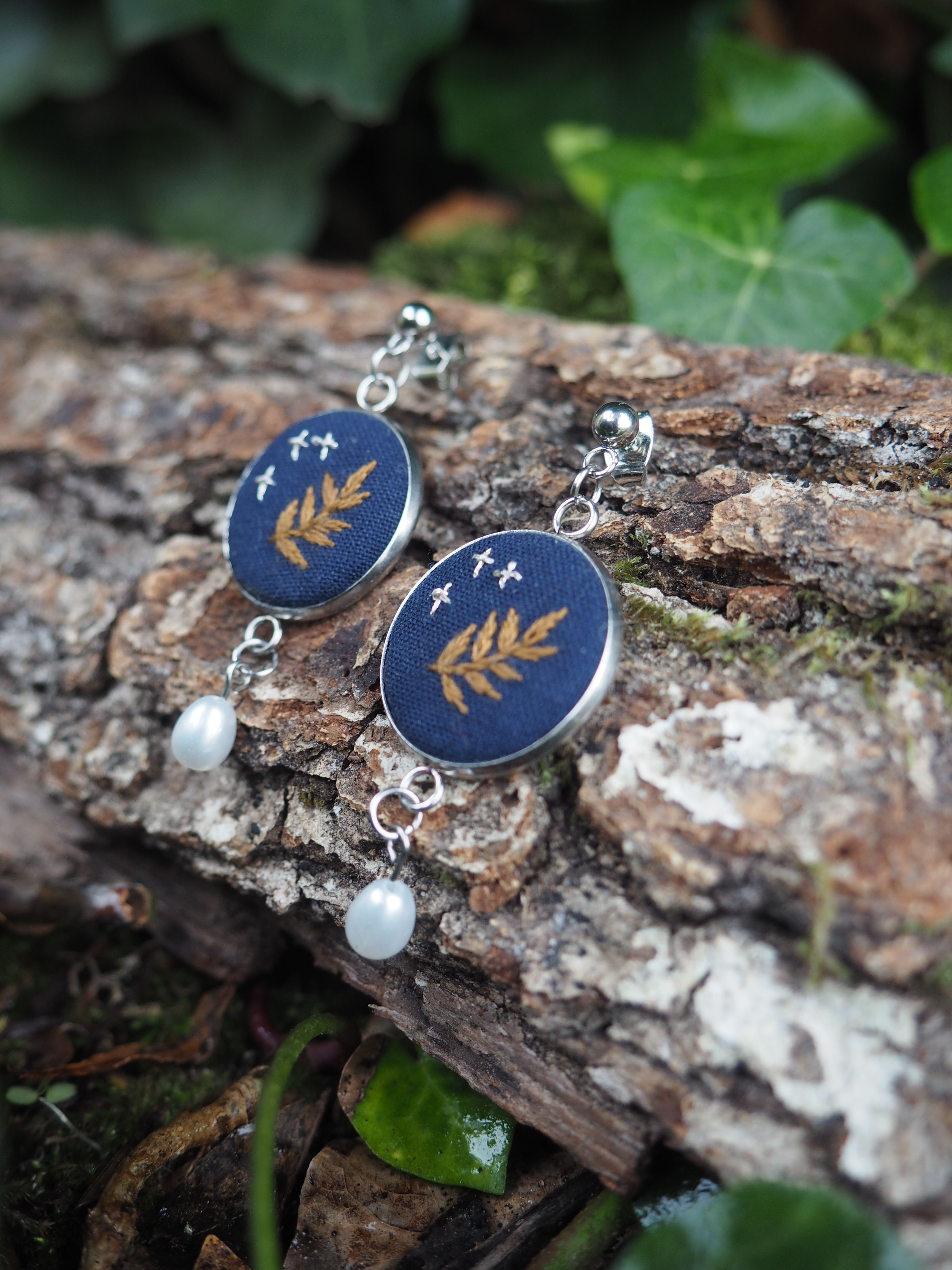 Cette image présente une paire de boucles d'oreilles artisanales disposées sur un morceau d'écorce dans un environnement naturel.  **Caractéristiques des bijoux :**
- Pendants circulaires de couleur bleu marine/indigo
- Motifs décoratifs dorés représentant des branches ou épis stylisés
- Petites étoiles dorées dispersées sur le fond bleu
- Perles blanches nacrées suspendues en pendentif
- Crochets d'oreilles en métal argenté **Mise en scène :**
- Support naturel : écorce d'arbre avec texture rugueuse et taches de lichen blanc
- Arrière-plan flou avec feuillage vert
- Éclairage naturel doux qui met en valeur les couleurs et textures
- Composition photographique soignée créant un contraste harmonieux entre l'artisanat et la nature Le style évoque un travail artisanal contemporain avec des influences botaniques, alliant élégance et simplicité dans un esprit "fait main" ou "nature".