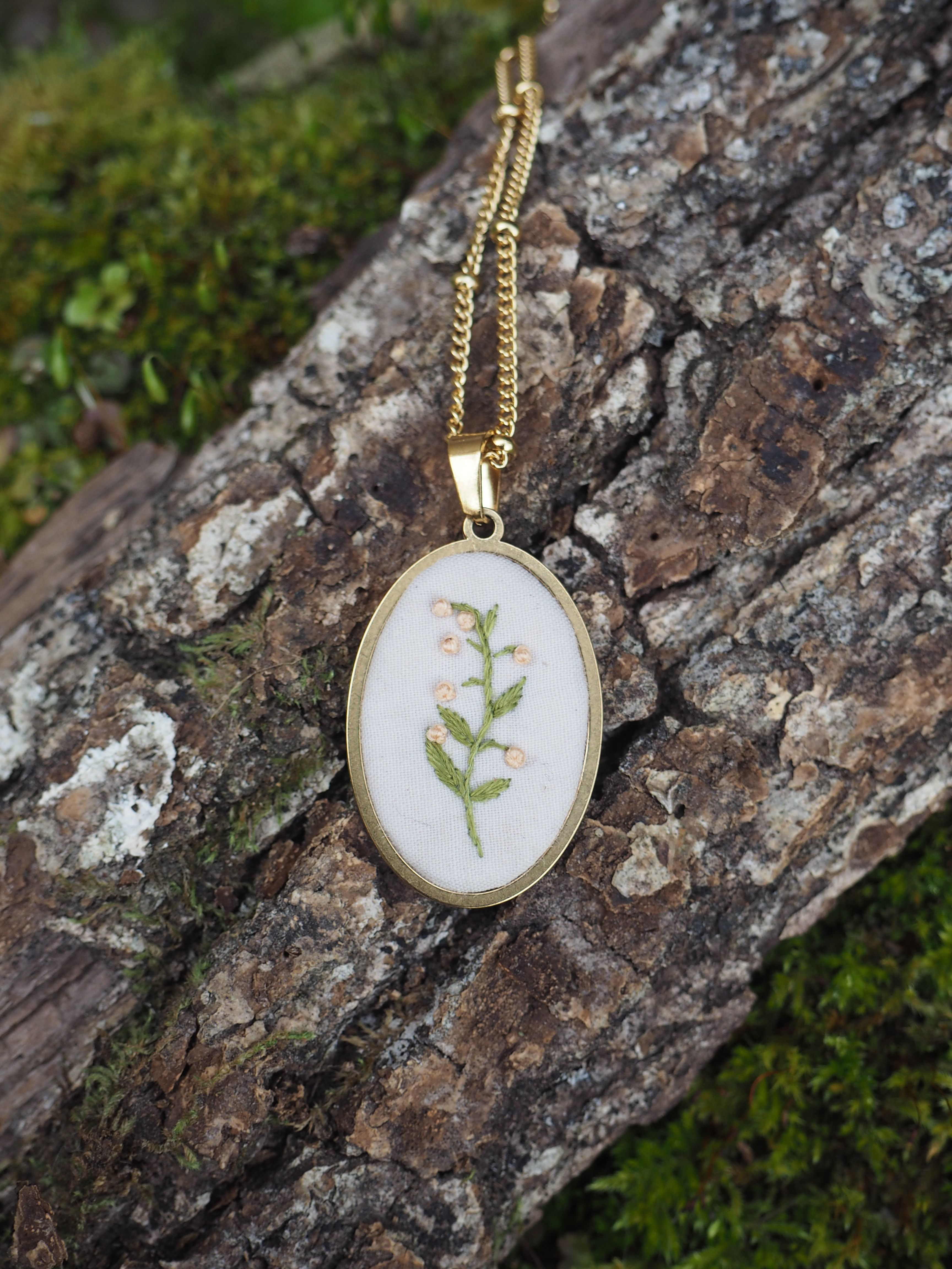 Cette image montre un délicat pendentif artisanal sur une chaîne dorée. Le pendentif est composé d'un cercle en métal doré qui encadre une broderie ou une impression botanique sur fond blanc. Le motif représente une petite fougère ou une brindille végétale verte, créant un effet naturel et délicat. Le bijou est photographié sur ce qui semble être un morceau de bois ou d'écorce recouvert de mousse, ce qui crée une mise en scène naturelle qui complimente bien le thème botanique du pendentif. L'ensemble dégage une atmosphère naturelle et poétique.