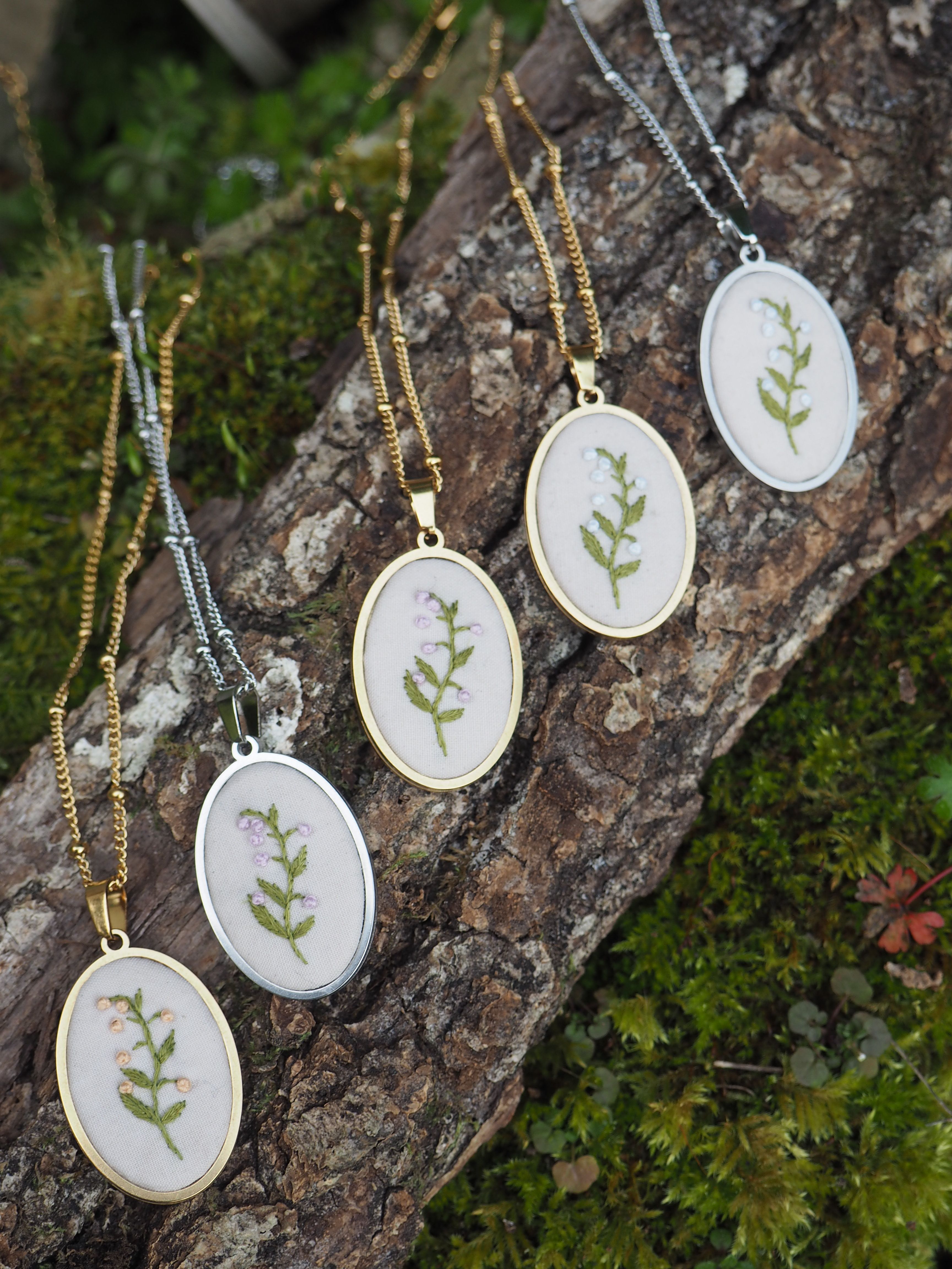Cette image montre une collection de délicats pendentifs brodés. Chaque pendentif est composé d'un petit cercle à broderie contenant un motif végétal simple - une petite branche verte avec des feuilles - encadré dans un support métallique rond. Les pendentifs sont présentés sur ce qui semble être un morceau d'écorce d'arbre ou de bois recouvert de mousse, créant un cadre naturel et organique. Les chaînes sont disponibles en deux finitions : dorée et argentée. L'ensemble dégage une atmosphère naturelle et botanique, parfaite pour les amateurs de bijoux inspirés par la nature.