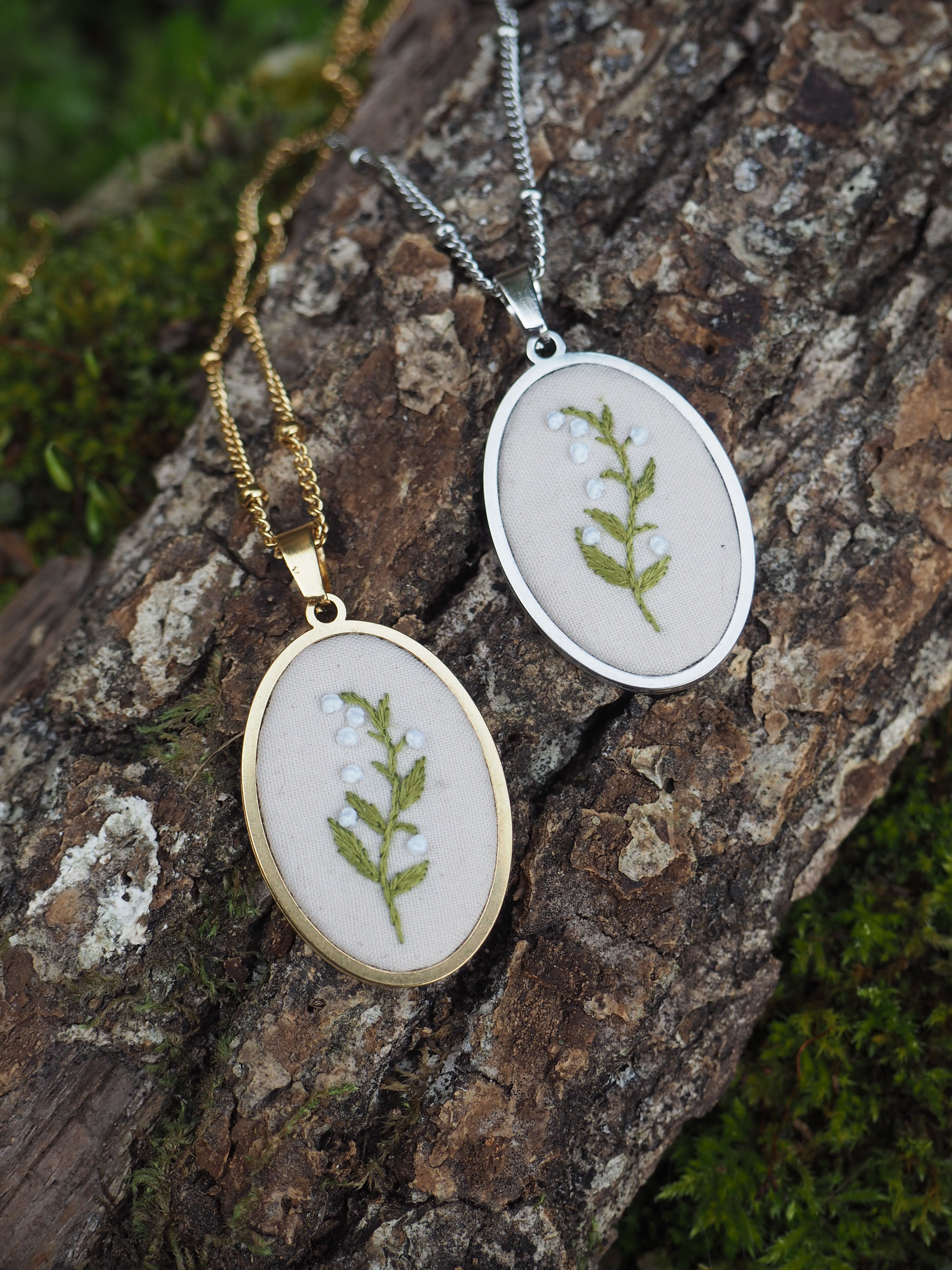 Cette image montre deux pendentifs délicats posés sur un morceau d'écorce d'arbre moussue. Les pendentifs sont de forme ovale, l'un en finition dorée et l'autre en finition argentée. Chacun contient une petite fougère ou une feuille verte pressée et préservée sous ce qui semble être du verre ou une résine transparente. Les pendentifs sont suspendus à des chaînes fines assorties à leur couleur respective (dorée et argentée). C'est un bel exemple de bijoux naturels qui incorporent des éléments botaniques, créant une pièce élégante et organique. La texture rugueuse de l'écorce et la mousse verte en arrière-plan complètent parfaitement ces bijoux nature.