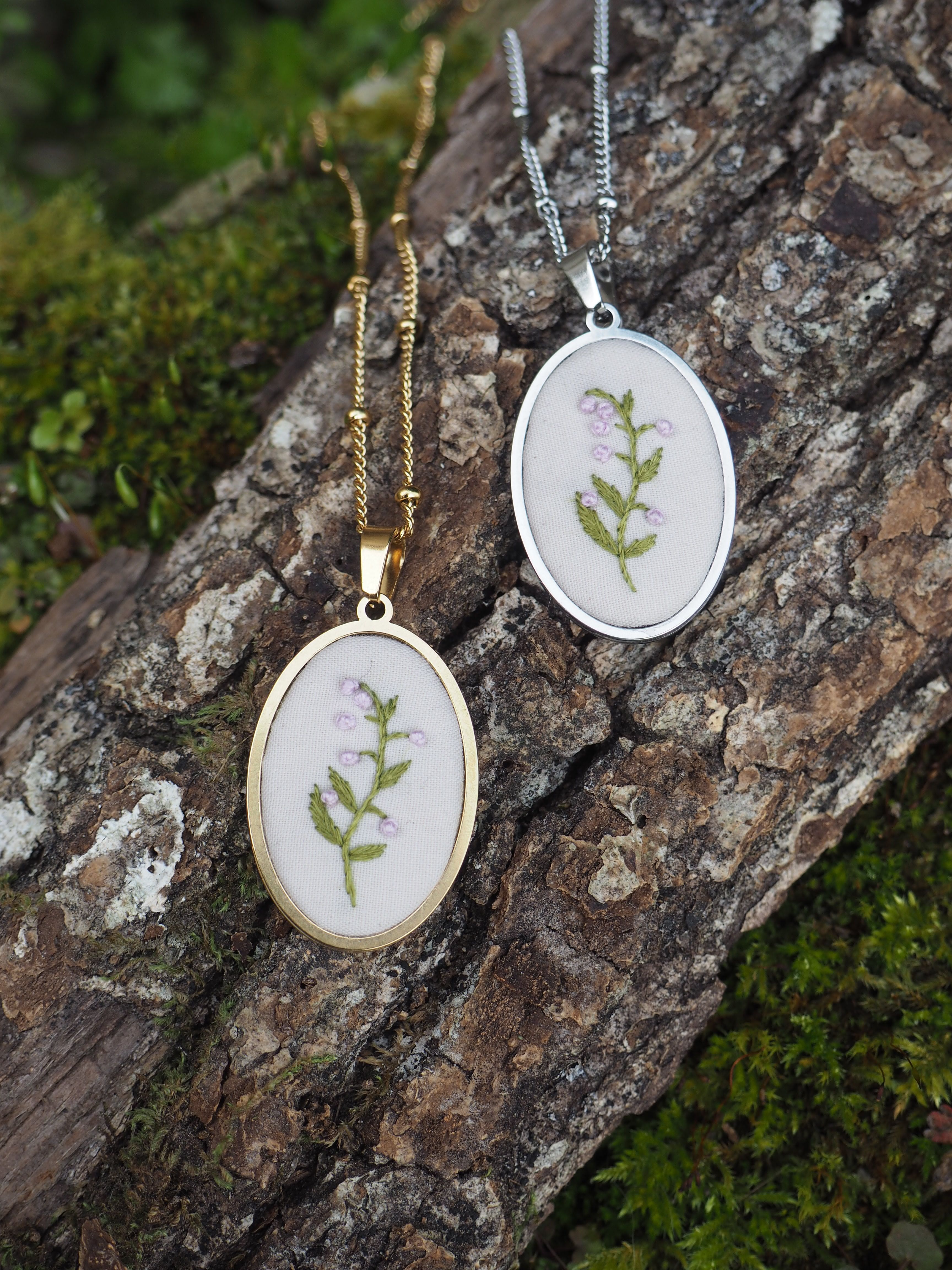 Cette image montre deux beaux pendentifs délicats posés sur un morceau d'écorce d'arbre moussue. Les pendentifs sont des médaillons circulaires, l'un en finition dorée et l'autre en finition argentée. Chacun contient une petite fougère ou une feuille verte pressée et préservée sous ce qui semble être un verre ou une résine transparente. Les pendentifs sont attachés à des chaînes assorties dans leurs tons respectifs (or et argent). La composition est très naturelle et organique, avec le contraste entre les bijoux raffinés et la texture rugueuse de l'écorce et la mousse verte en arrière-plan. Ces pendentifs représentent un bel exemple de bijoux botaniques qui incorporent des éléments naturels dans leur design.