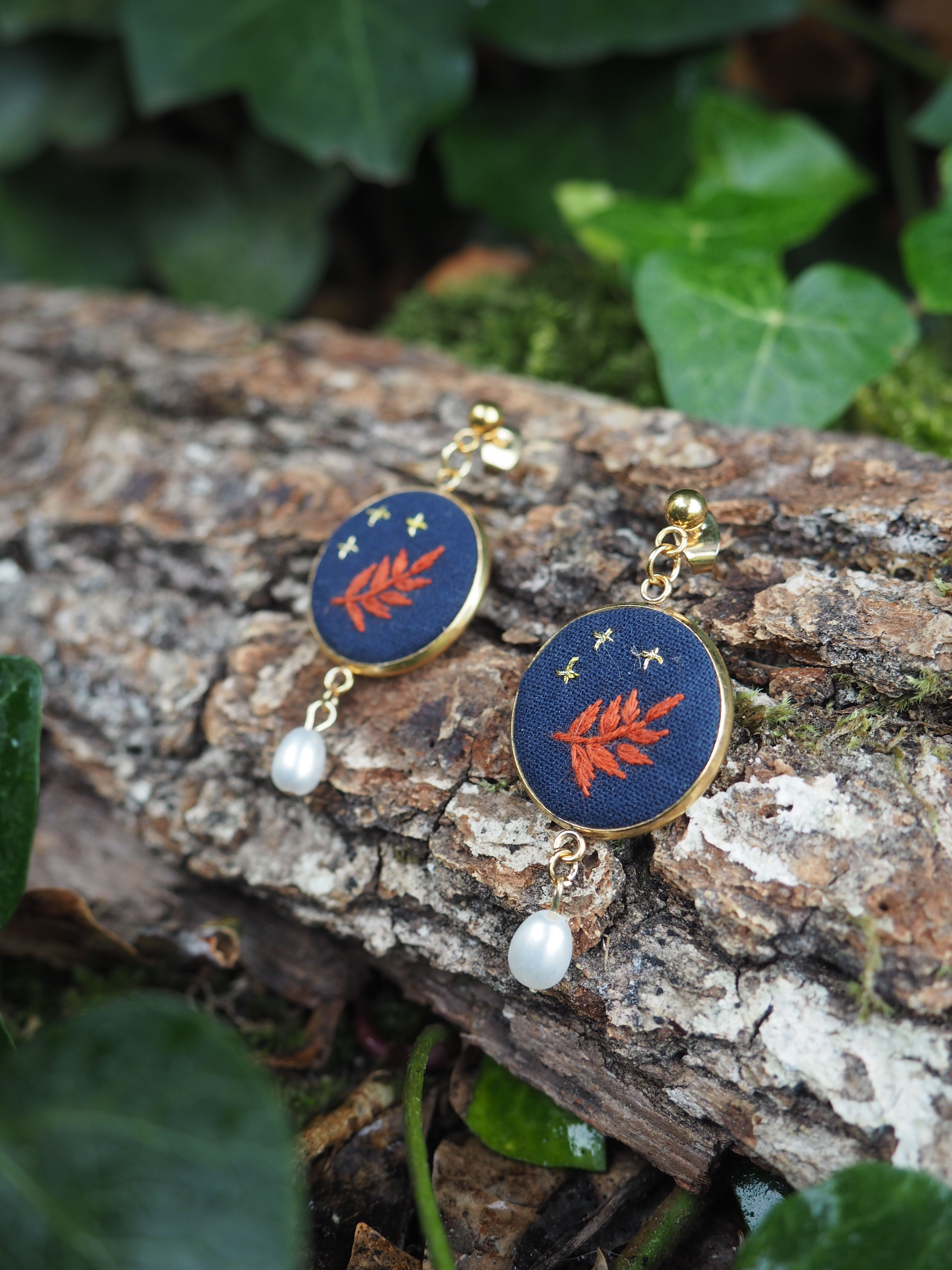 Cette image présente une paire de boucles d'oreilles élégantes posées sur un morceau de bois naturel dans un environnement verdoyant. **Description des bijoux :**
- Boucles d'oreilles composées de médaillons circulaires à fond bleu marine
- Motif décoratif : une feuille ou branche stylisée de couleur rouge-orangé avec de petites étoiles dorées dispersées
- Monture dorée qui encadre chaque médaillon
- Petites perles blanches nacrées suspendues en pendentif sous chaque médaillon
- Style raffiné mêlant couleurs vives et éléments naturels **Mise en scène :**
- Les bijoux reposent sur une surface de bois brut, avec une écorce texturée et patinée
- Arrière-plan composé de feuillage vert luxuriant, légèrement flou
- Éclairage naturel qui met en valeur les couleurs et les détails des boucles d'oreilles
- Composition harmonieuse entre les éléments naturels et les bijoux artisanaux L'ensemble crée une atmosphère organique et artisanale, suggérant des bijoux de créateur inspirés par la nature.