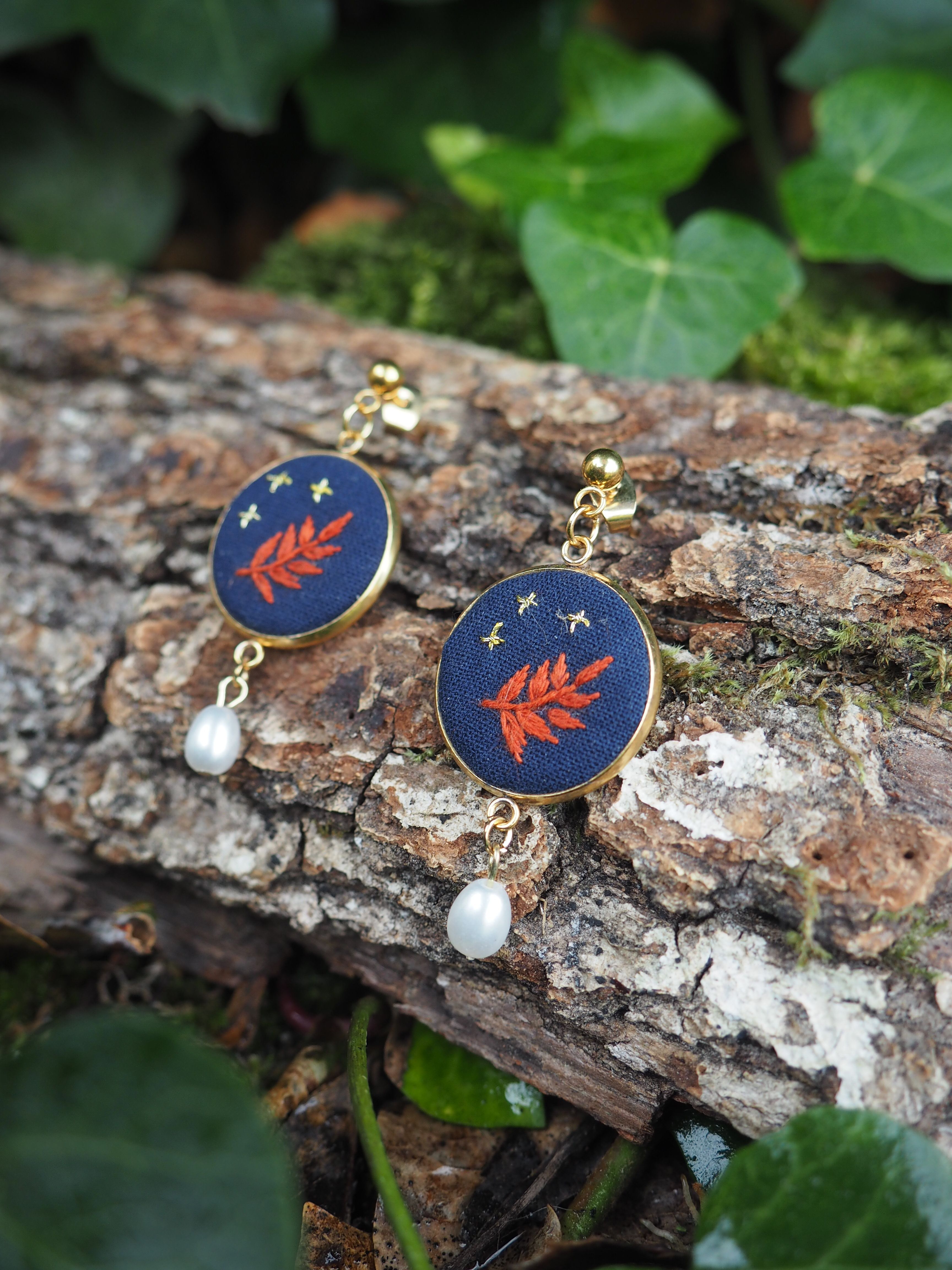 Cette image présente une paire de boucles d'oreilles artisanales disposées sur un tronc d'arbre dans un environnement naturel.  **Caractéristiques des bijoux :**
- Forme circulaire avec un fond bleu marine/navy
- Motif central représentant une feuille ou branche stylisée en couleur orange/corail
- Petites étoiles dorées dispersées sur le fond bleu
- Perle blanche nacrée suspendue à chaque boucle
- Crochets dorés pour l'attache **Environnement :**
- Les boucles d'oreilles reposent sur l'écorce rugueuse d'un tronc d'arbre
- Arrière-plan naturel avec un feuillage vert luxuriant légèrement flou
- Éclairage naturel qui met en valeur les couleurs et les détails
- Quelques traces de lichen ou mousse visible sur l'écorce Le style de ces bijoux évoque un design bohème ou artisanal, avec une esthétique qui rappelle un ciel étoilé nocturne orné d'éléments botaniques. La mise en scène naturelle renforce le caractère organique et authentique de ces accessoires.