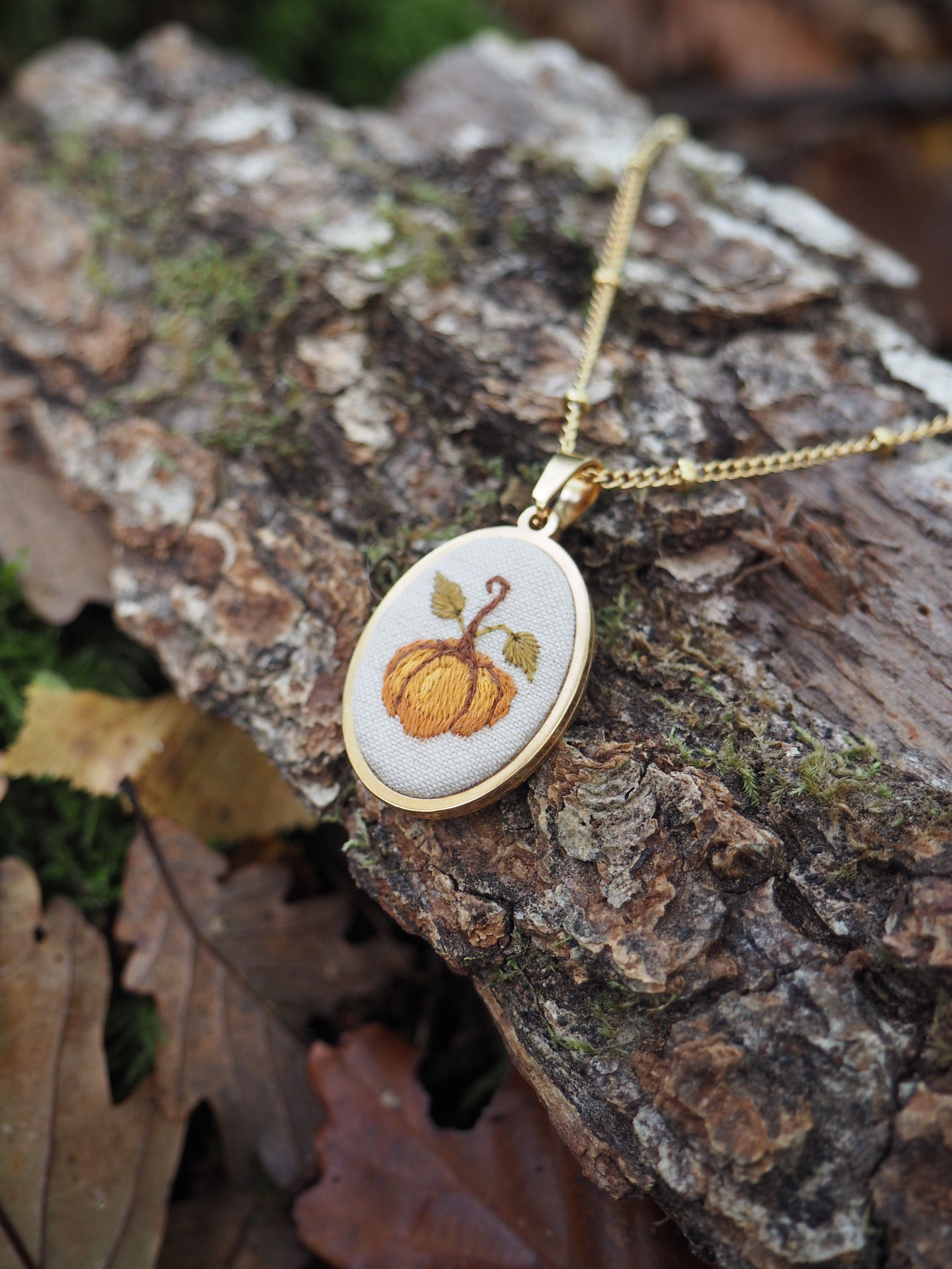 Cette image présente un délicat pendentif artisanal posé sur un morceau d'écorce d'arbre dans un environnement naturel. Le bijou se compose d'un petit cadre ovale en bois clair, probablement du bouleau ou un bois similaire, suspendu à une fine chaîne dorée. À l'intérieur du pendentif, on peut admirer une broderie minutieuse représentant une citrouille orange avec sa tige verte, réalisée sur un tissu de fond blanc ou crème. Les détails de la broderie sont particulièrement soignés, avec des nuances d'orange qui donnent du relief et de la texture au motif automnal. Le pendentif repose sur une surface d'écorce rugueuse et texturée, parsemée de mousse verdâtre, ce qui crée un contraste intéressant entre l'objet délicat et l'environnement naturel brut. En arrière-plan, on distingue des feuilles mortes aux tons bruns et cuivrés, renforçant l'atmosphère automnale de la composition. L'ensemble évoque un style artisanal fait main, probablement une pièce unique alliant broderie traditionnelle et joaillerie contemporaine, parfaite pour célébrer la saison d'automne.