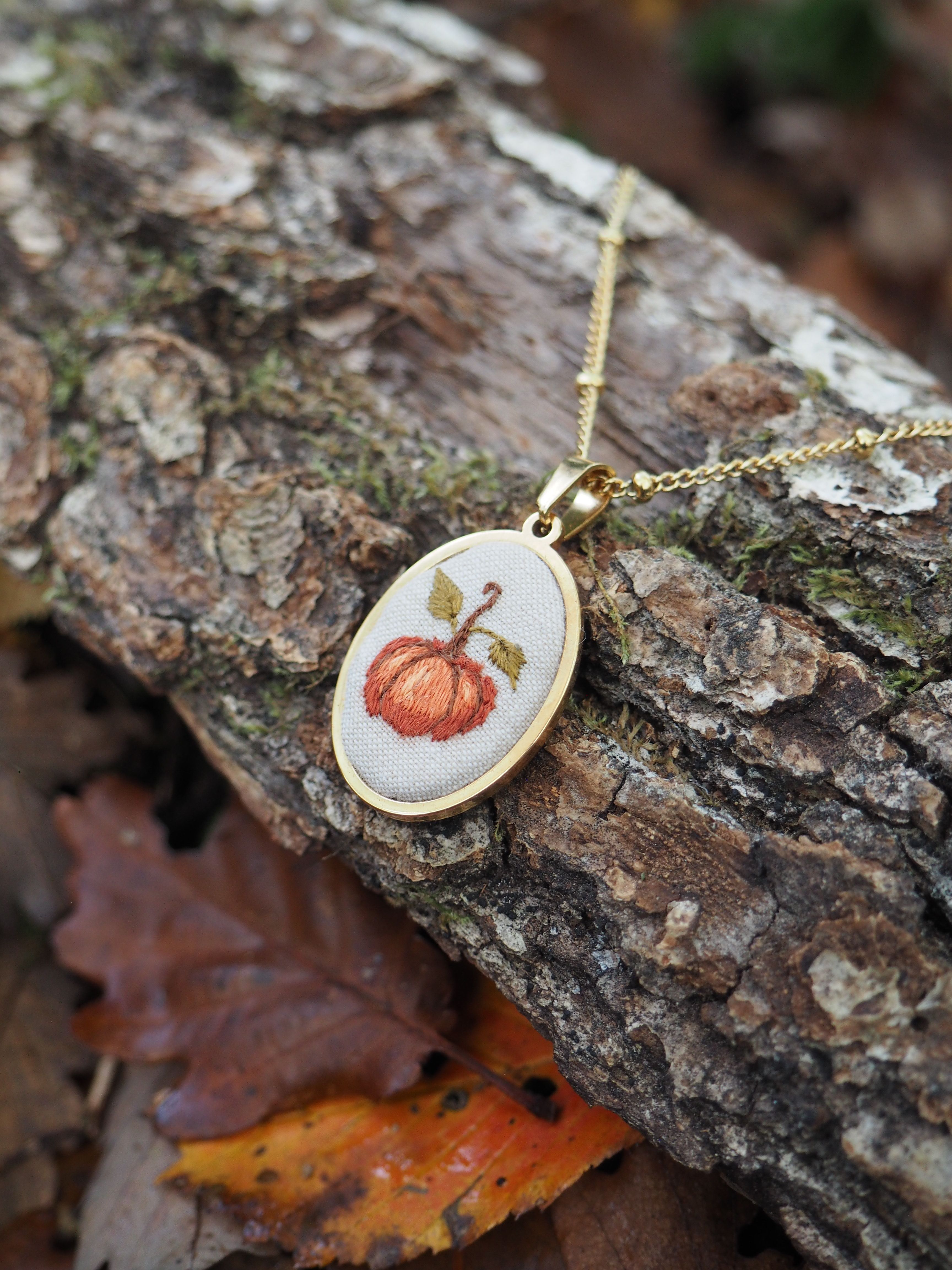 Cette image présente un délicat pendentif artisanal posé sur un morceau d'écorce d'arbre dans un environnement naturel automnal. Le bijou se compose d'un petit cadre ovale doré contenant une broderie minutieuse représentant une citrouille orange avec sa tige verte. Le travail de broderie est remarquablement détaillé, avec des points soignés qui donnent du relief et de la texture au motif. Le pendentif est suspendu à une fine chaîne dorée et repose sur l'écorce rugueuse et texturée d'un tronc d'arbre, dont on peut voir les nuances de brun et les traces de mousse. En arrière-plan, on aperçoit des feuilles mortes aux teintes automnales - orange, brun et jaune - qui jonchent le sol, créant une atmosphère saisonnière parfaitement en harmonie avec le motif de citrouille du bijou. L'ensemble évoque l'artisanat fait main, la saison d'automne et la beauté des petits détails décoratifs inspirés de la nature. La composition photographique met en valeur le contraste entre la délicatesse du travail de broderie et la rusticité de l'environnement naturel.