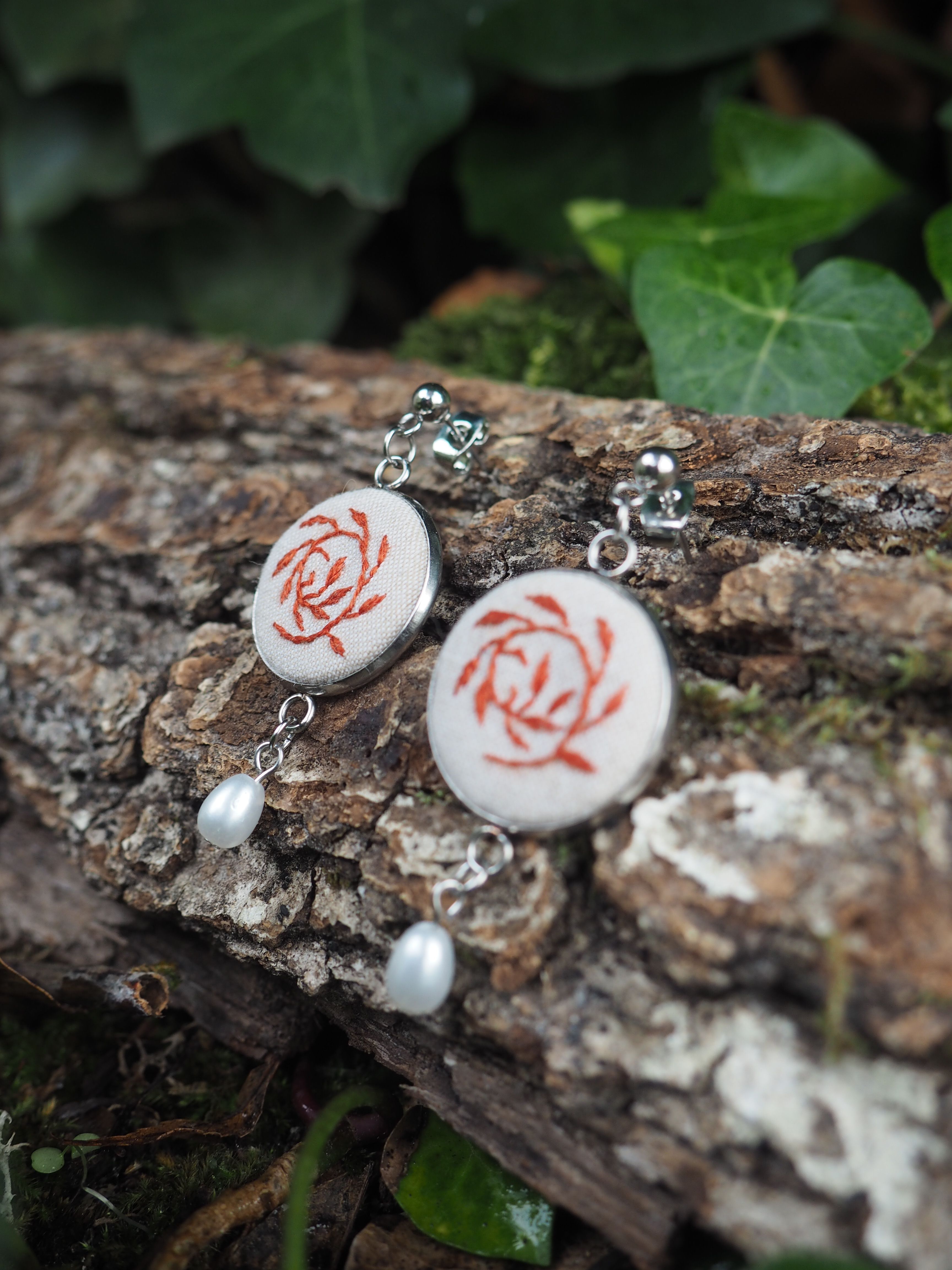 Cette image présente une paire de boucles d'oreilles artisanales disposées sur un tronc d'arbre dans un environnement naturel.  **Caractéristiques des bijoux :**
- Pendants circulaires en céramique ou porcelaine blanche
- Motif floral rouge/rose représentant des roses stylisées
- Montures en métal argenté avec des crochets d'oreilles
- Petites perles blanches nacrées suspendues en pendentif
- Style romantique et délicat **Mise en scène :**
- Posés sur l'écorce rugueuse et texturée d'un tronc d'arbre
- Arrière-plan naturel avec un feuillage vert flou
- Éclairage naturel doux qui met en valeur les détails
- Composition photographique soignée créant un contraste entre la délicatesse des bijoux et la rusticité du support en bois L'ensemble évoque un style artisanal fait main, avec une esthétique à la fois naturelle et raffinée, parfait pour un look bohème ou romantique.