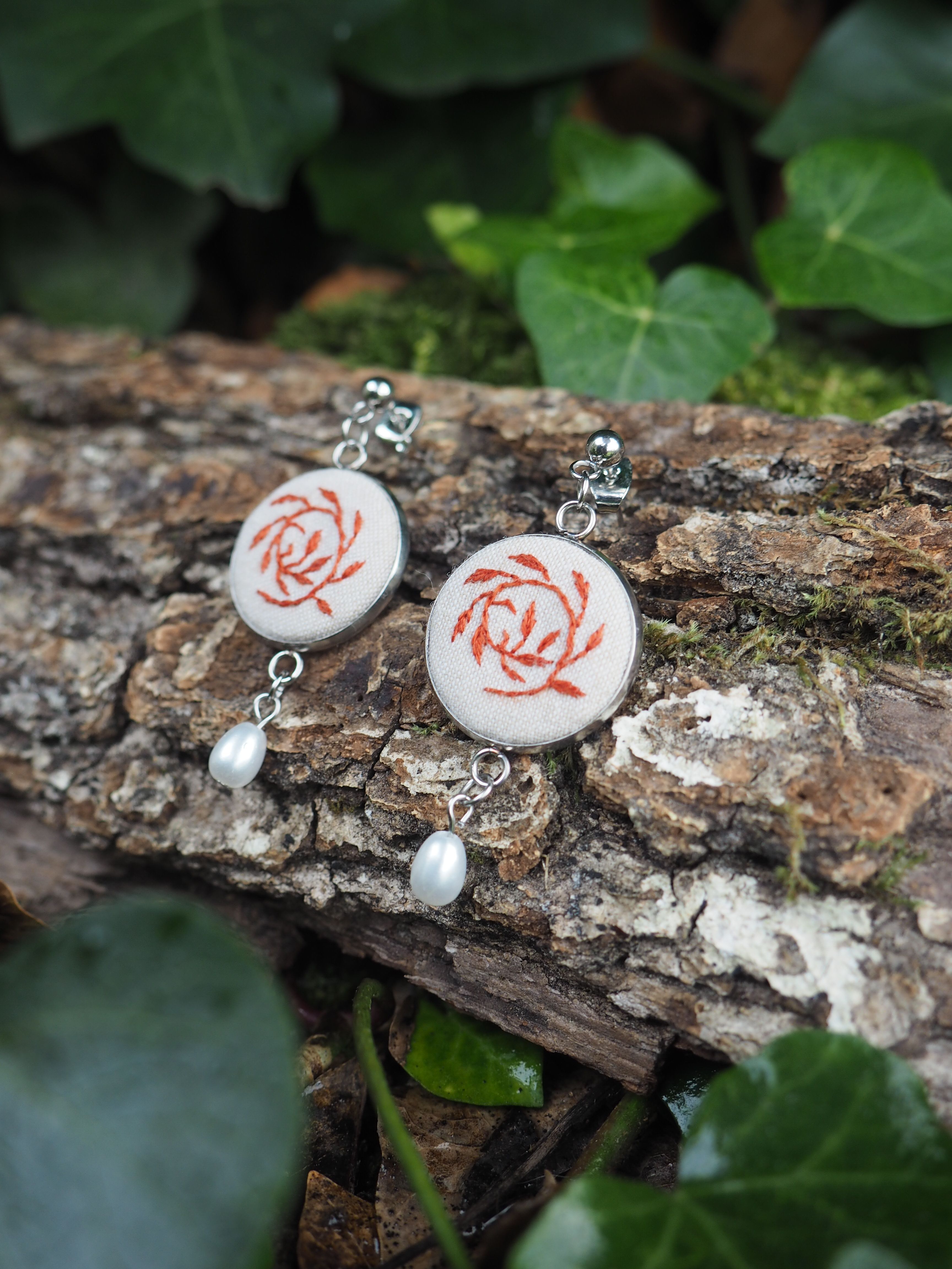 Cette image présente une paire de boucles d'oreilles artisanales disposées sur un morceau de bois naturel dans un environnement végétal. **Description détaillée :** **Les boucles d'oreilles :**
- Composées de pendentifs circulaires en céramique ou porcelaine blanche
- Ornées d'un motif décoratif rouge/rose en forme de spirale ou de rose stylisée
- Surmontées de crochets métalliques argentés
- Agrémentées de petites perles blanches nacrées suspendues en pendentif **Le support :**
- Morceau de bois flotté ou branche naturelle avec une texture rugueuse
- Surface patinée et vieillie, présentant des nuances de brun et de gris
- Quelques traces de mousse ou de lichen ajoutent au caractère naturel **L'environnement :**
- Arrière-plan composé de feuillage vert luxuriant
- Feuilles de différentes tailles créant un bokeh naturel
- Ambiance forestière ou de jardin sauvage
- Éclairage naturel doux qui met en valeur les bijoux Cette mise en scène crée un contraste harmonieux entre l'artisanat délicat des bijoux et la rusticité de l'environnement naturel, soulignant le caractère fait-main et authentique des boucles d'oreilles.