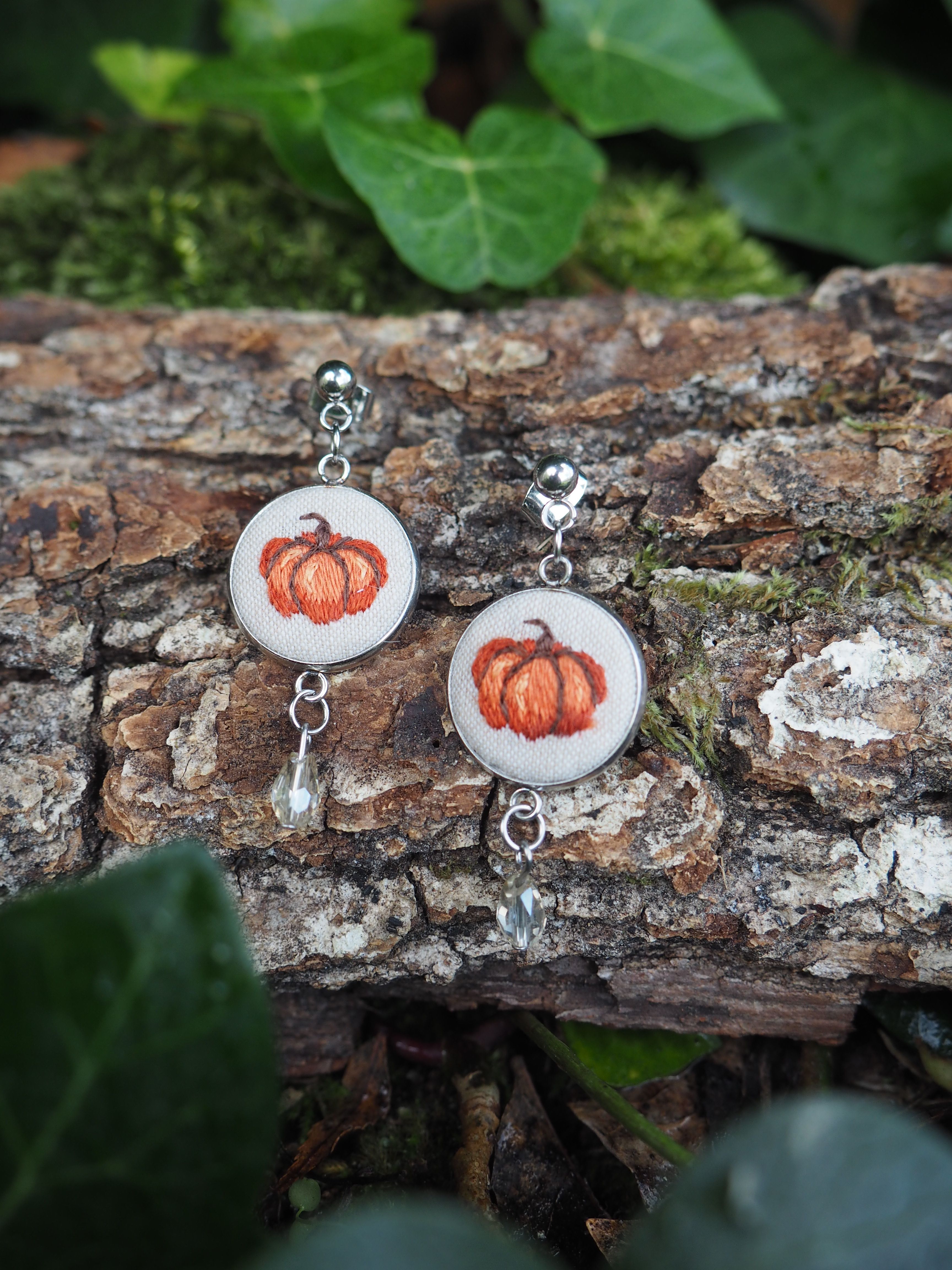 Cette image présente une paire de boucles d'oreilles artisanales disposées sur une surface naturelle en bois. Les bijoux sont composés d'éléments circulaires blancs ou crème sur lesquels sont peints ou imprimés des motifs de citrouilles orange, évoquant l'automne ou Halloween.  Les boucles d'oreilles sont ornées de petites perles ou breloques pendantes qui ajoutent du mouvement et de l'élégance à l'ensemble. Les crochets d'oreilles semblent être en métal argenté. L'arrière-plan naturel, constitué d'écorce d'arbre texturée et de feuillage vert flou, crée un contraste harmonieux qui met en valeur les bijoux. Cette mise en scène extérieure donne une ambiance organique et artisanale à la photographie, suggérant que ces accessoires sont probablement faits main et s'inspirent de thèmes saisonniers. L'éclairage naturel et la composition soignée mettent bien en évidence les détails et les couleurs des boucles d'oreilles.