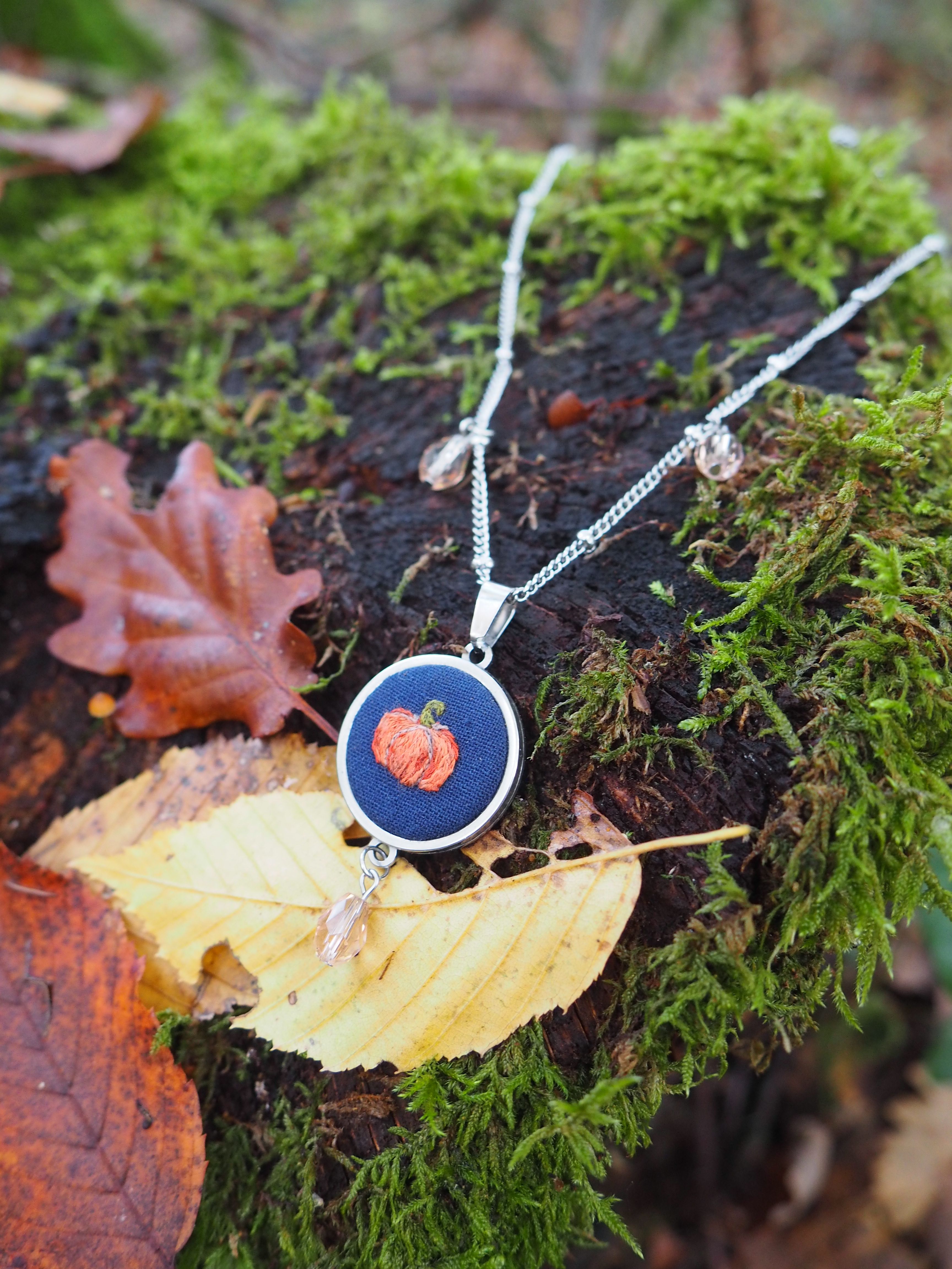 Cette image présente un bijou artisanal photographié dans un cadre naturel très évocateur. Il s'agit d'un pendentif circulaire suspendu à une chaîne argentée, posé délicatement sur ce qui semble être un champignon ou un morceau de bois clair. Le pendentif lui-même est particulièrement intéressant : il présente un fond bleu profond sur lequel se détache un motif central de couleur rouge-orangé, possiblement une fleur ou un élément décoratif naturel. Le contraste entre ces couleurs chaudes et froides crée un effet visuel saisissant. L'environnement choisi pour cette mise en scène est remarquable : le bijou repose sur un lit de mousse verte luxuriante, parsemée de feuilles d'automne aux teintes cuivrées et rougeâtres. Cette composition évoque parfaitement l'atmosphère forestière et automnale, créant une harmonie parfaite entre l'objet artisanal et son environnement naturel. L'éclairage doux et la profondeur de champ utilisée mettent en valeur les détails du pendentif tout en créant un arrière-plan naturellement flou qui renforce l'aspect contemplatif de l'image.