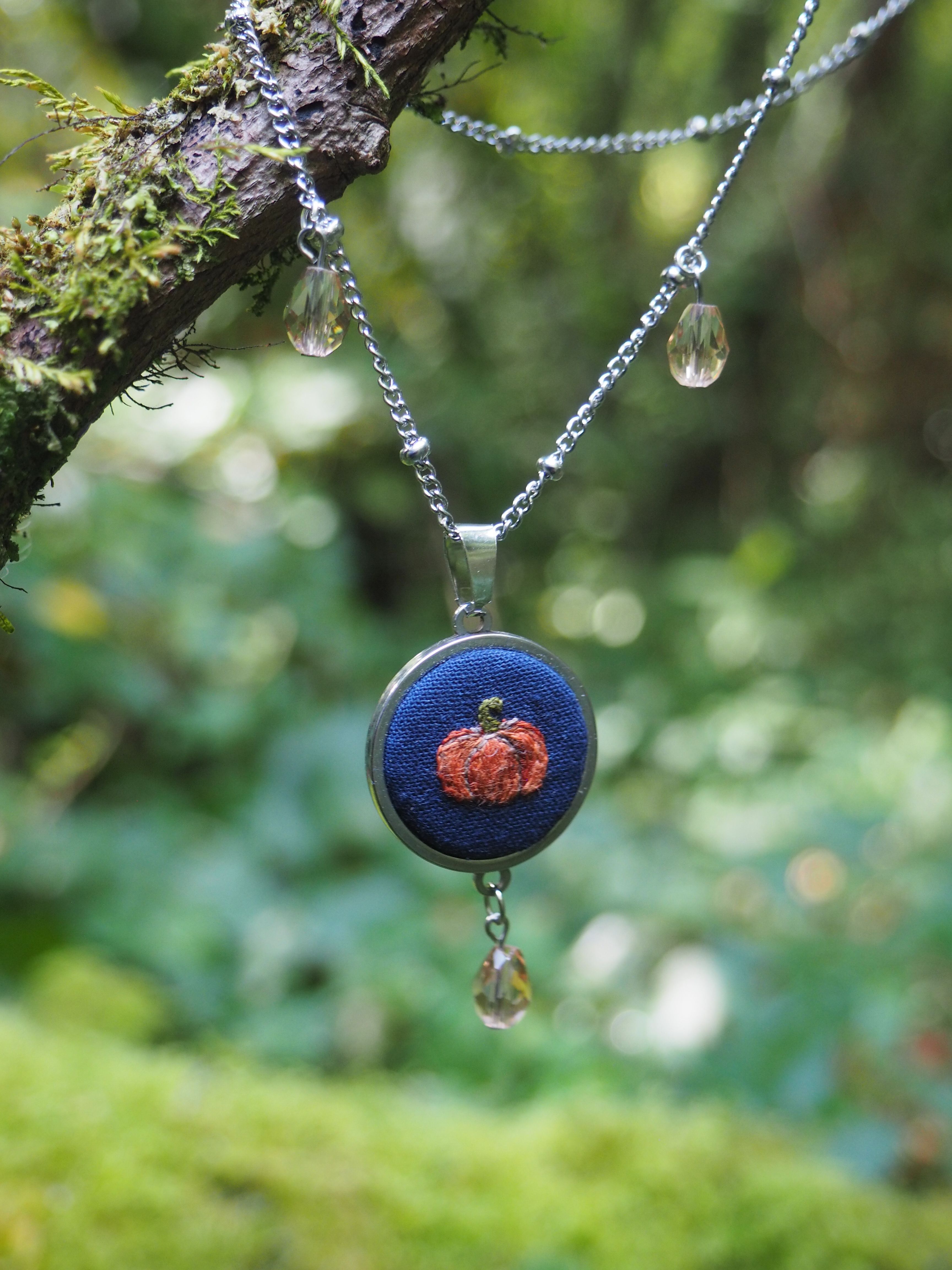 Cette image présente un magnifique collier artisanal suspendu à une branche d'arbre dans un environnement naturel luxuriant. **Éléments principaux :** - **Pendentif central** : Une pièce ovale avec un fond bleu profond orné d'un motif floral rouge/orange au centre, probablement réalisé en technique de broderie ou d'incrustation
- **Chaîne** : Fine chaîne argentée ou métallique qui serpente élégamment
- **Perles décoratives** : Plusieurs petites perles transparentes ou cristallines suspendues à différents points de la chaîne **Environnement :**
- Branche d'arbre couverte de mousse et de lichen, témoignant d'un écosystème humide
- Arrière-plan naturel avec un bokeh vert doux créé par la végétation floue
- Lumière naturelle douce qui met en valeur les détails du bijou **Esthétique :**
L'ensemble dégage une atmosphère bohème et naturelle, où l'artisanat humain s'harmonise parfaitement avec l'environnement forestier. La composition photographique crée un contraste saisissant entre les couleurs vives du pendentif et les tons verts apaisants de la nature environnante.