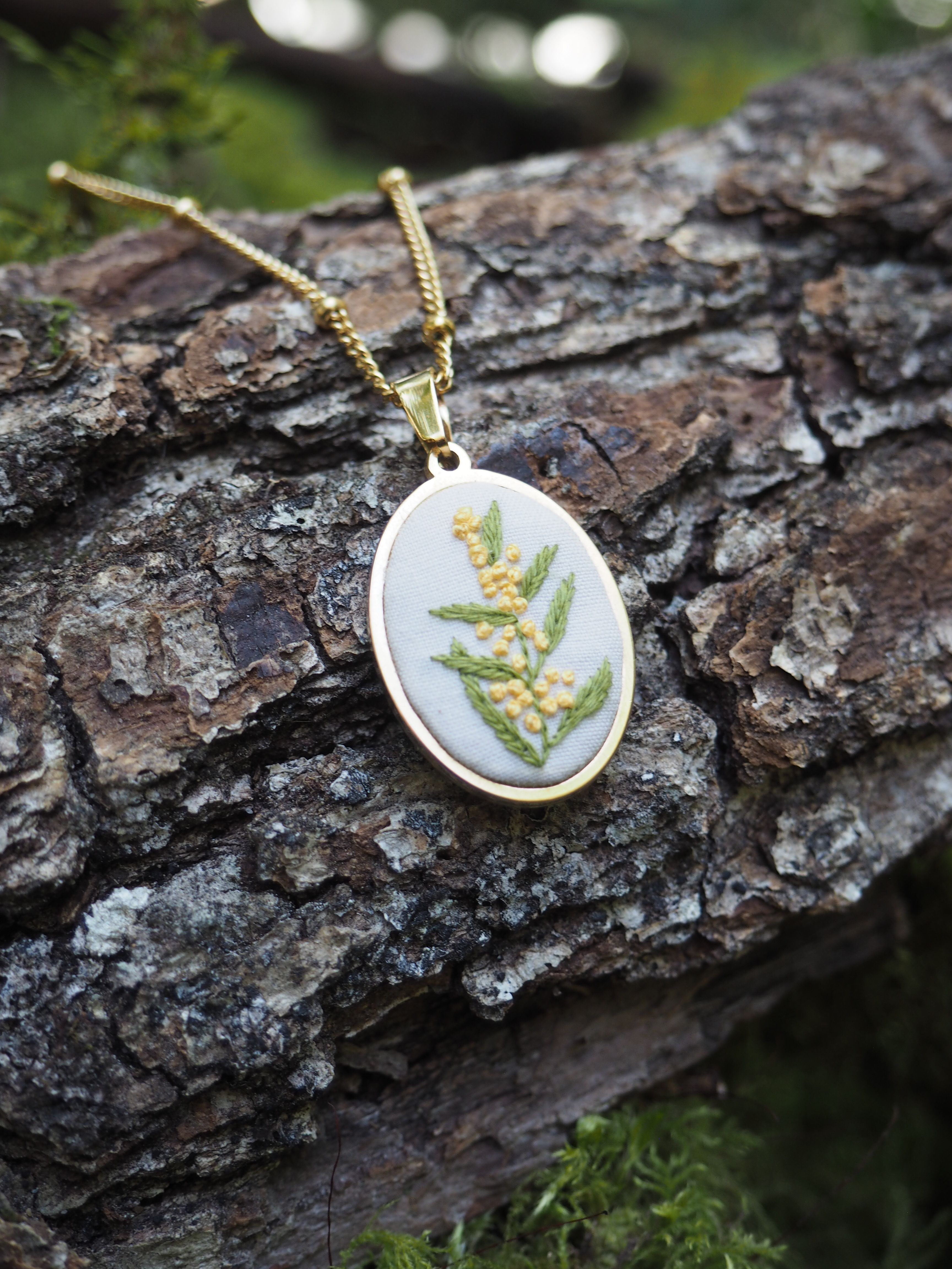 Cette image montre un délicat pendentif sur une chaîne dorée, photographié sur un morceau d'écorce d'arbre texturée. Le pendentif est de forme ovale ou ronde et présente un motif de fougère verte pressée ou peinte sur un fond blanc. C'est une pièce de bijouterie nature très élégante qui capture la beauté de la botanique. Le contraste entre le pendentif délicat et l'écorce rugueuse crée une belle composition, avec un peu de mousse verte visible dans le cadre, ajoutant à l'ambiance naturelle de la photo.