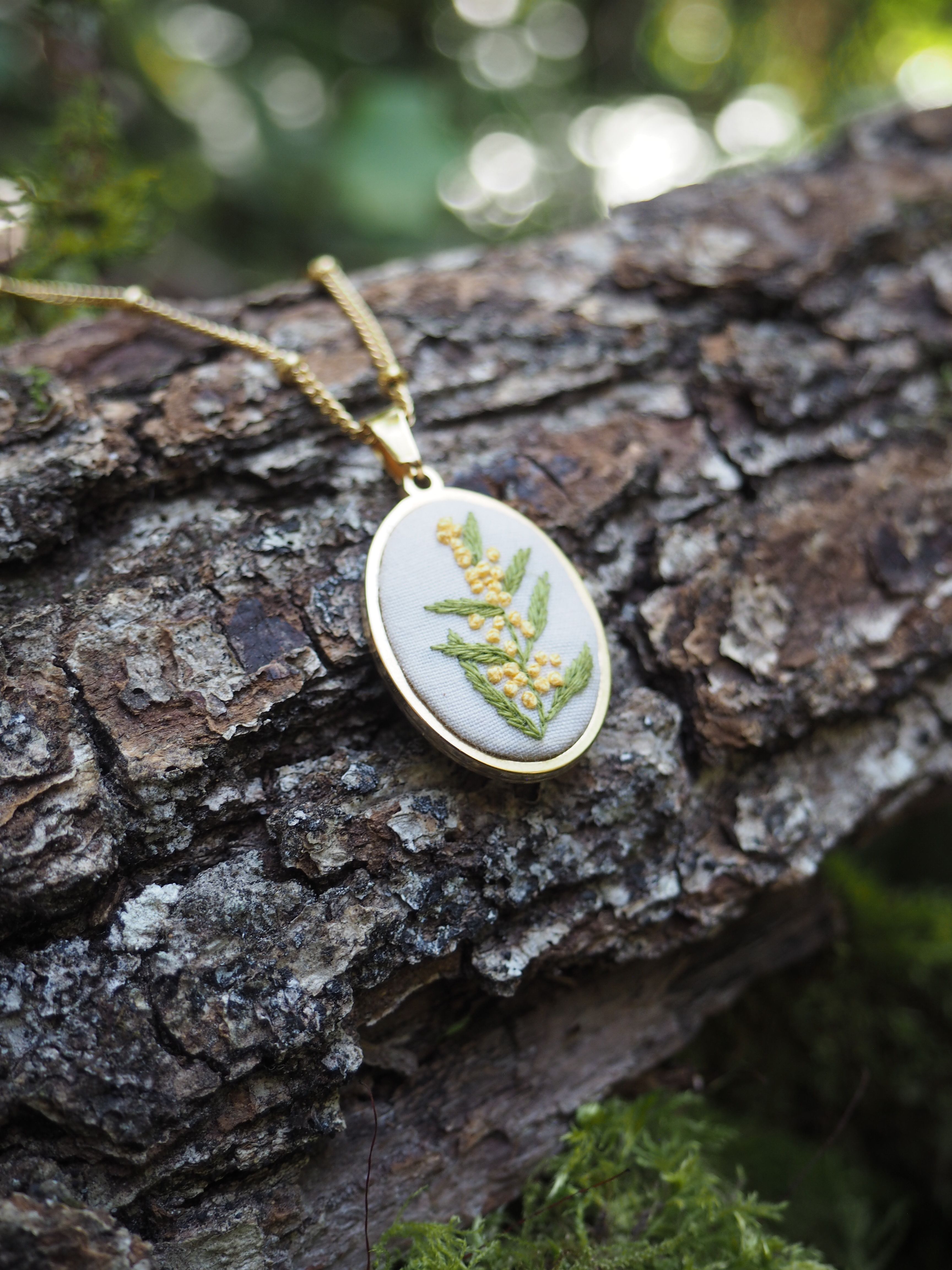 Cette image montre un délicat pendentif ovale posé sur un tronc d'arbre à l'écorce rugueuse. Le pendentif semble être fait d'argent ou d'un métal argenté, et contient de véritables feuilles ou fougères pressées et préservées sous verre ou résine. Il est suspendu à une chaîne dorée. L'arrière-plan est flou mais on peut distinguer des touches de vert et de la lumière naturelle créant un effet bokeh. C'est une belle composition qui met en valeur ce bijou artisanal inspiré par la nature, photographié dans un cadre naturel qui fait écho à ses motifs botaniques.