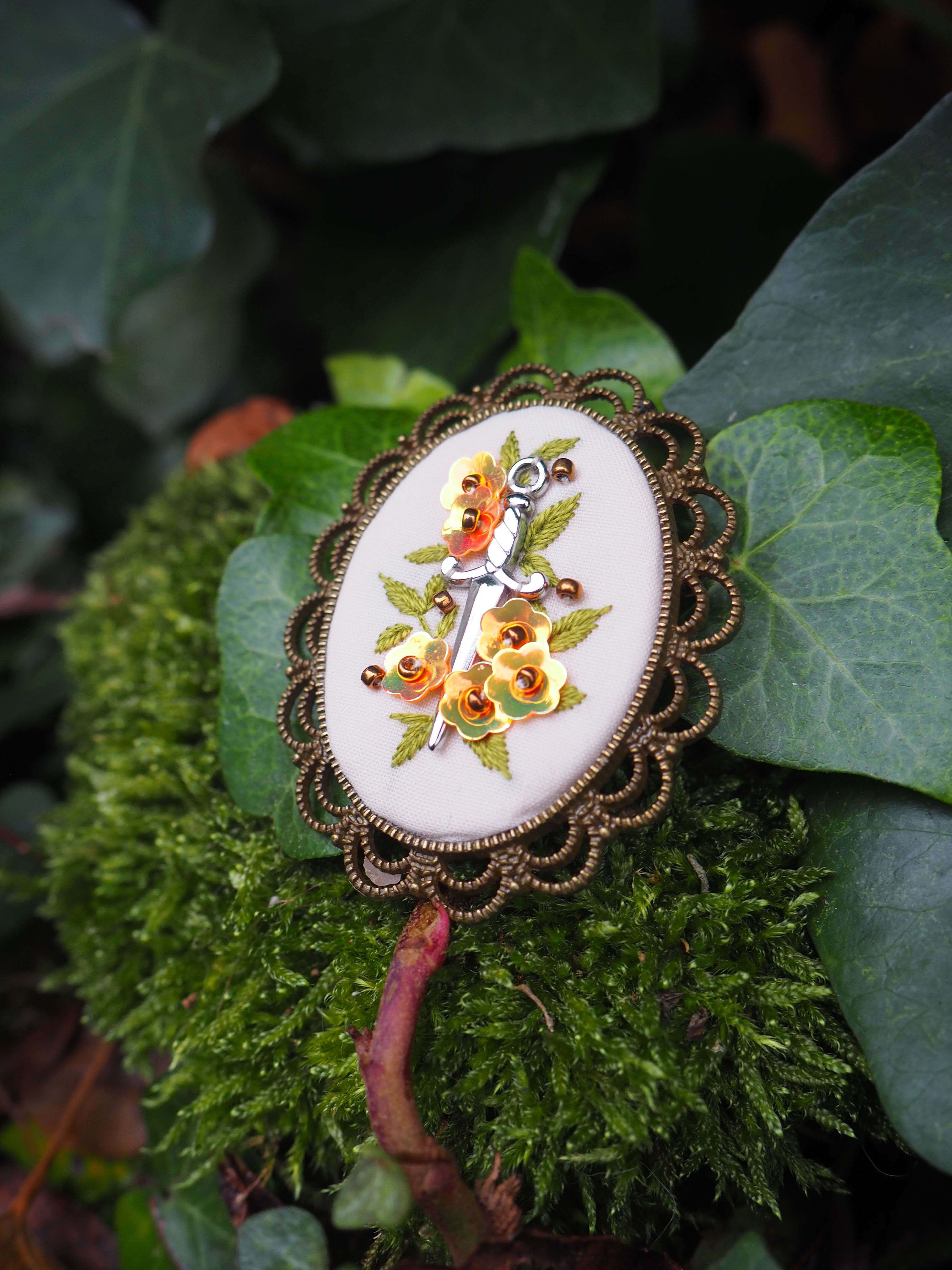 Cette image présente une broche ou un pendentif décoratif posé sur un lit de mousse verte dans un environnement naturel. La pièce de bijouterie présente les caractéristiques suivantes : **Design et structure :**
- Forme ovale avec un contour dentelé élégant de couleur bronze ou cuivre
- Centre en émail ou porcelaine blanche
- Motif floral peint à la main représentant des roses ou pivoines dans des tons chauds (orange, rouge, jaune)
- Style vintage ou rétro, évoquant les bijoux de la fin du 19ème ou début du 20ème siècle **Environnement :**
- Posé délicatement sur de la mousse verte luxuriante
- Entouré de feuilles naturelles aux nuances vertes variées
- Éclairage doux qui met en valeur les détails du motif floral
- Ambiance naturelle et organique qui contraste harmonieusement avec l'aspect raffiné du bijou Cette mise en scène crée un effet artistique saisissant, où l'élégance de l'objet manufacturé se marie parfaitement avec la beauté brute de la nature environnante.