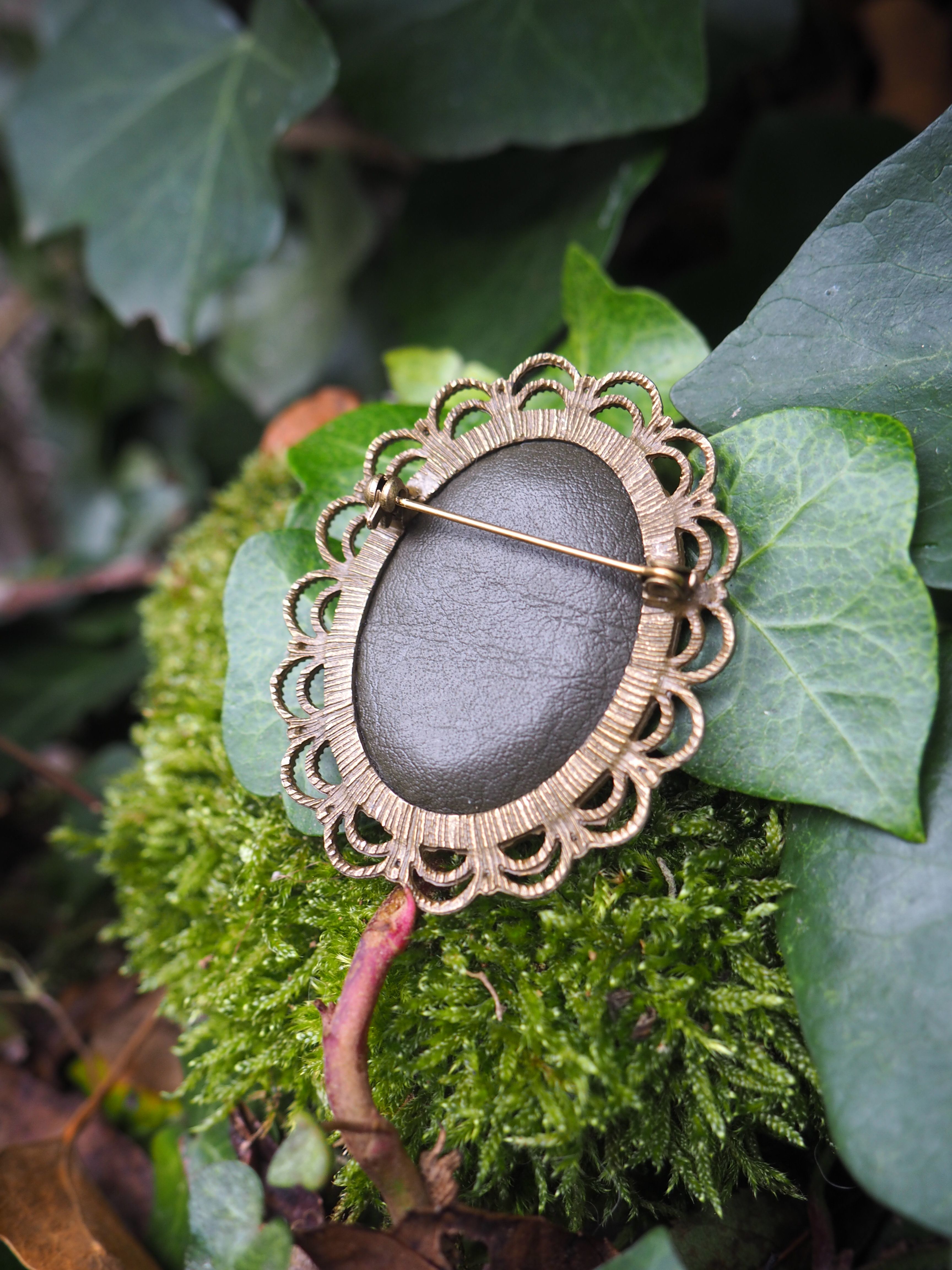 Cette image présente un bijou artisanal élégant posé sur un lit de mousse verte dans un environnement naturel. Il s'agit d'une broche ou d'un pendentif de forme ovale, caractérisé par : **Le design :**
- Une pierre centrale lisse de couleur gris-mauve aux reflets nacrés
- Un sertissage en métal doré (probablement du laiton ou du bronze) travaillé en forme de pétales ou de festons décoratifs
- Une épingle ou attache visible sur le côté **L'environnement :**
- Le bijou repose sur de la mousse d'un vert vif et dense
- Il est entouré de feuilles vertes de différentes tailles et textures
- L'arrière-plan présente un feuillage naturel légèrement flou
- L'éclairage naturel met en valeur les détails du travail artisanal **L'esthétique :**
Ce bijou évoque un style bohème ou vintage, avec son travail métallique finement ciselé qui rappelle les créations artisanales. La mise en scène dans cet écrin de verdure souligne le caractère naturel et organique de la pièce, créant une harmonie entre l'objet façonné et son environnement végétal.