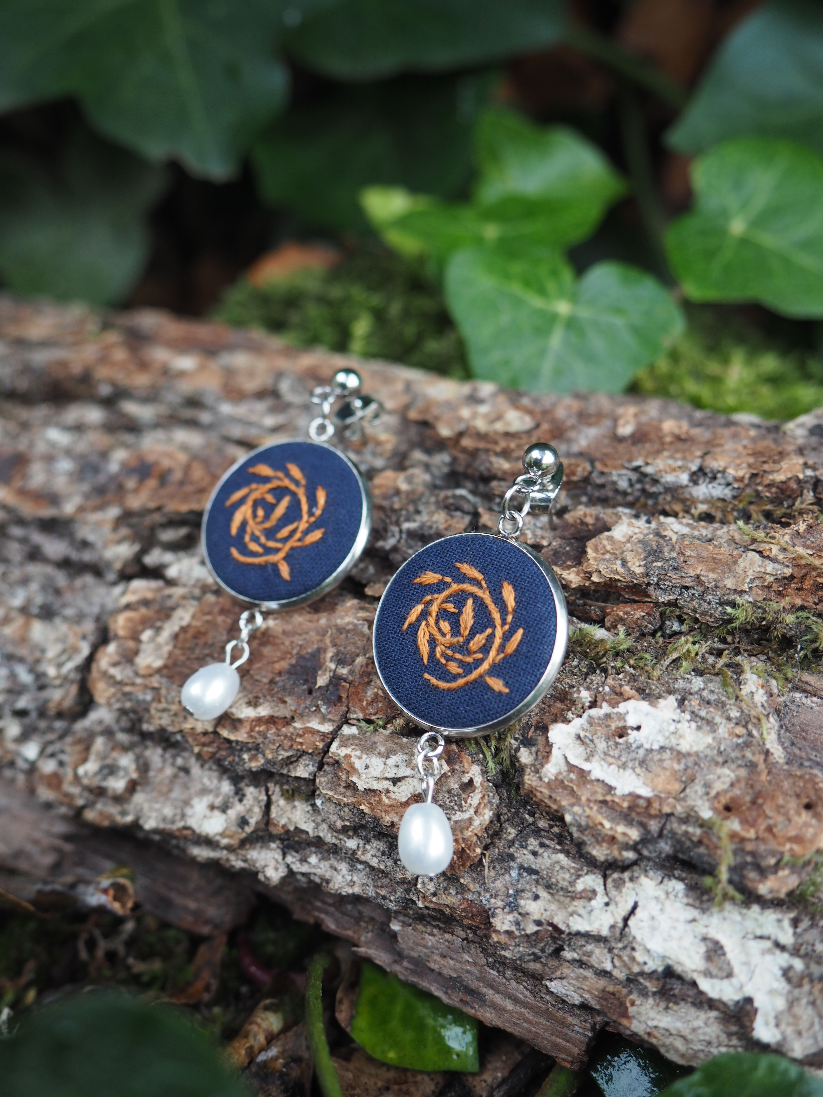 Cette image présente une paire de boucles d'oreilles artisanales disposées sur un tronc d'arbre dans un environnement naturel.  **Caractéristiques des bijoux :**
- Pendants circulaires de couleur bleu marine/indigo
- Motif décoratif doré au centre ressemblant à un nœud celtique ou à une rosace stylisée
- Petites perles blanches nacrées suspendues en dessous
- Crochets d'oreilles en métal argenté **Environnement :**
- Support : écorce rugueuse d'un tronc d'arbre avec des lichens blancs
- Arrière-plan : feuillage vert flou créant un bokeh naturel
- Éclairage doux et naturel qui met en valeur les couleurs des bijoux **Composition photographique :**
- Mise au point nette sur les boucles d'oreilles
- Profondeur de champ réduite pour isoler le sujet
- Contraste harmonieux entre les tons bleus des bijoux et les verts de la végétation Cette présentation met en valeur le caractère artisanal et naturel de ces bijoux, suggérant un style bohème ou ethnique.