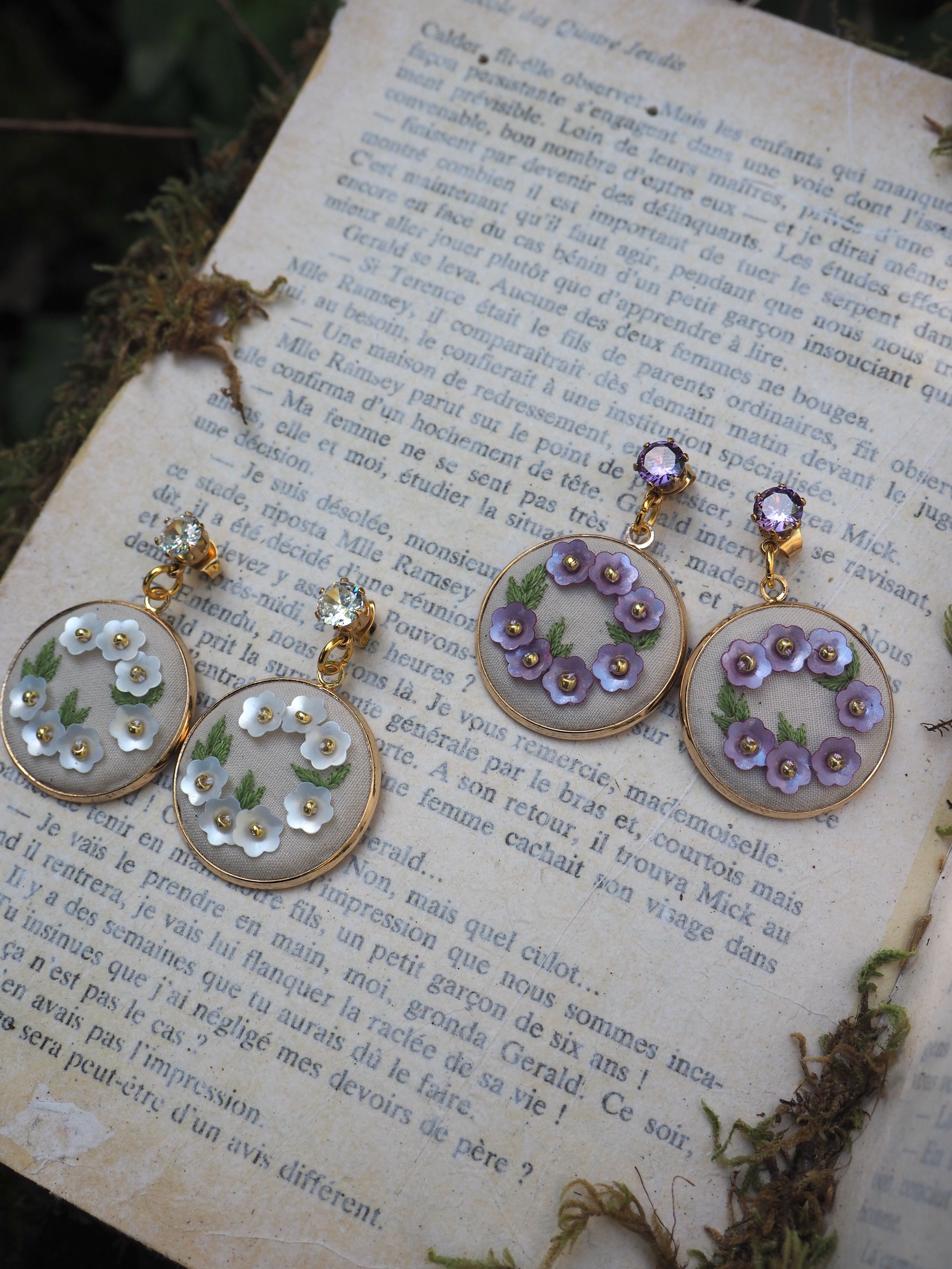 Ces belles boucles d'oreilles artisanales sont photographiées sur une page de livre ancien en français. Les bijoux semblent être composés de cercles transparents ornés de petites fleurs délicates - une paire avec des fleurs blanches/vertes et l'autre avec des fleurs violettes. Les cercles paraissent être en résine ou en verre, montés sur des supports dorés. Les fleurs miniatures donnent un aspect très romantique et vintage à ces bijoux. La mise en scène sur la page vieillie du livre ajoute une touche poétique et nostalgique à la photo.