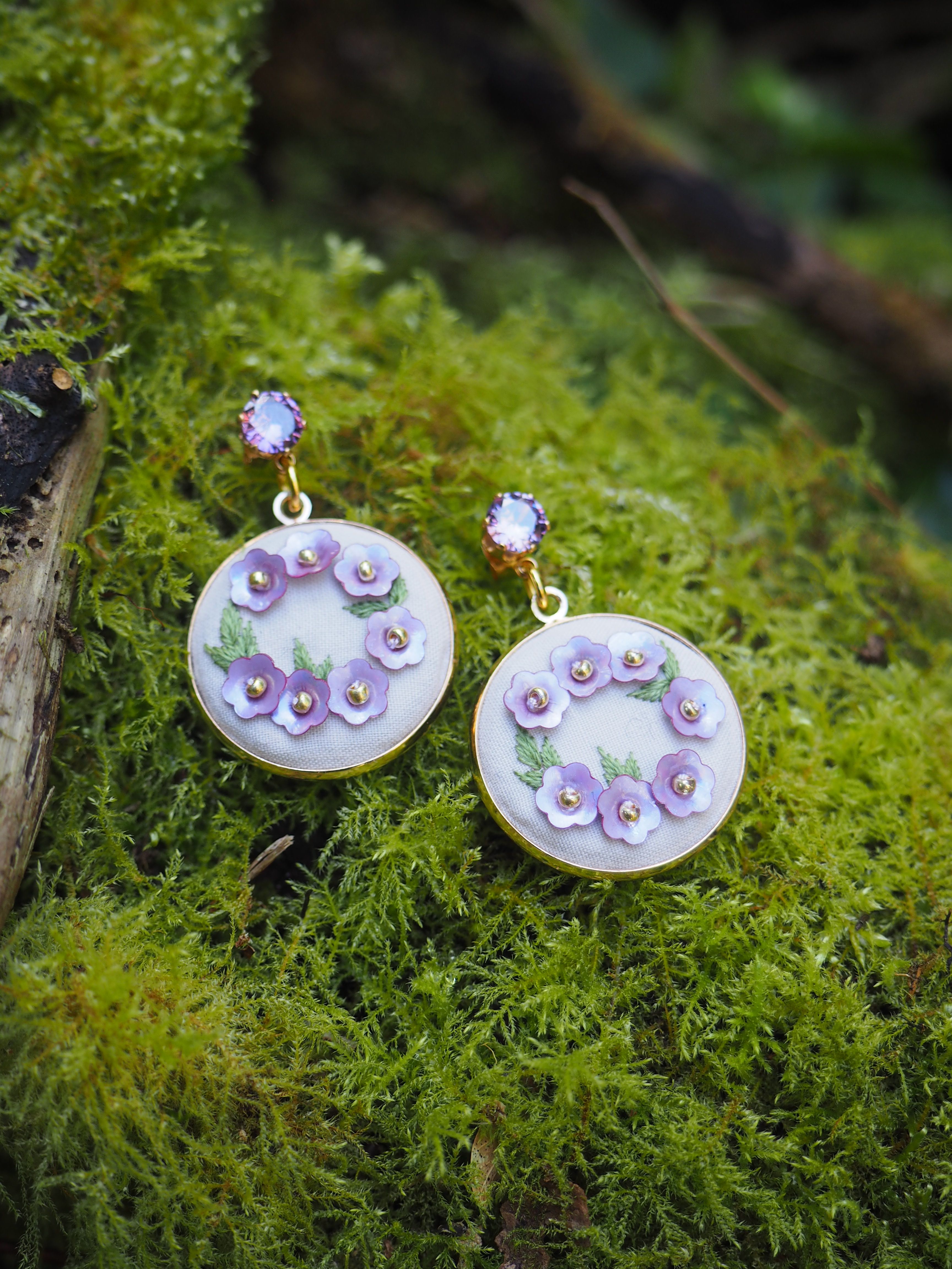Cette image montre une paire de boucles d'oreilles artisanales posées sur un lit de mousse verte. Les boucles d'oreilles sont de forme circulaire, avec un fond blanc ou nacré, et sont décorées de petites fleurs délicates en tons de violet et lavande. Les fleurs semblent être disposées en cercle sur chaque pendentif. Les boucles d'oreilles ont également ce qui semble être des petits cristaux ou perles au centre des fleurs. Le contraste entre les bijoux délicats et la mousse naturelle verte crée une belle composition photographique qui évoque une ambiance féerique ou enchantée.