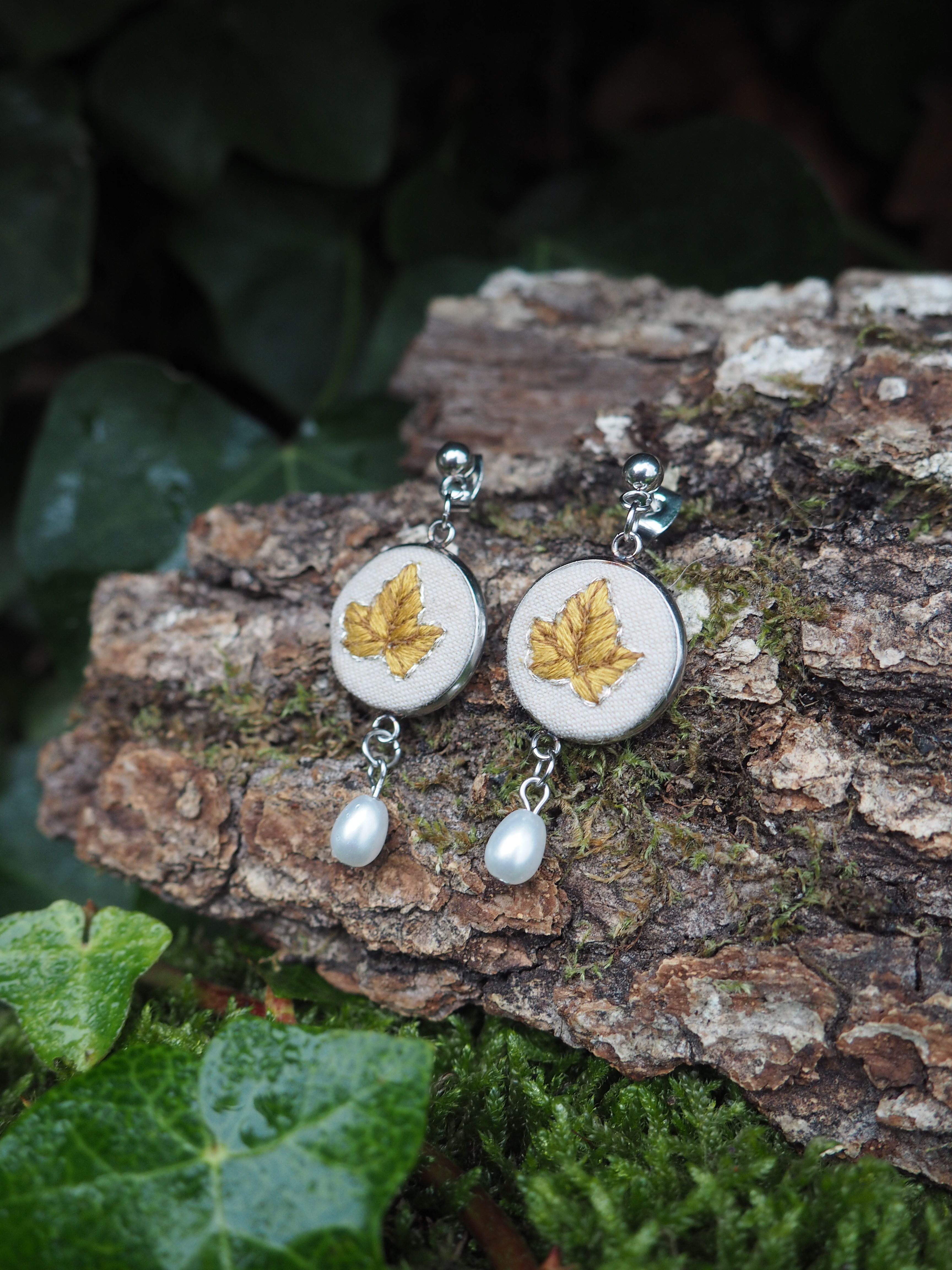 This image features a pair of elegant earrings resting on a piece of bark, surrounded by lush greenery. The earrings are silver-toned and dangle. Each earring consists of a circular, flat disc with a light, possibly off-white or cream-colored fabric surface. On this surface, a detailed golden leaf motif is embroidered or painted, resembling a maple or sycamore leaf with visible veins. Below each disc, a single, lustrous white pearl hangs, adding a touch of classic sophistication. The bark on which the earrings are placed is rough and textured, with shades of brown and hints of green moss, suggesting an outdoor or natural setting. In the foreground and background, vibrant green ivy leaves and other foliage are visible, creating a natural and earthy backdrop that complements the delicate design of the earrings. The lighting appears soft, highlighting the details of the jewelry and the natural textures of the surroundings.