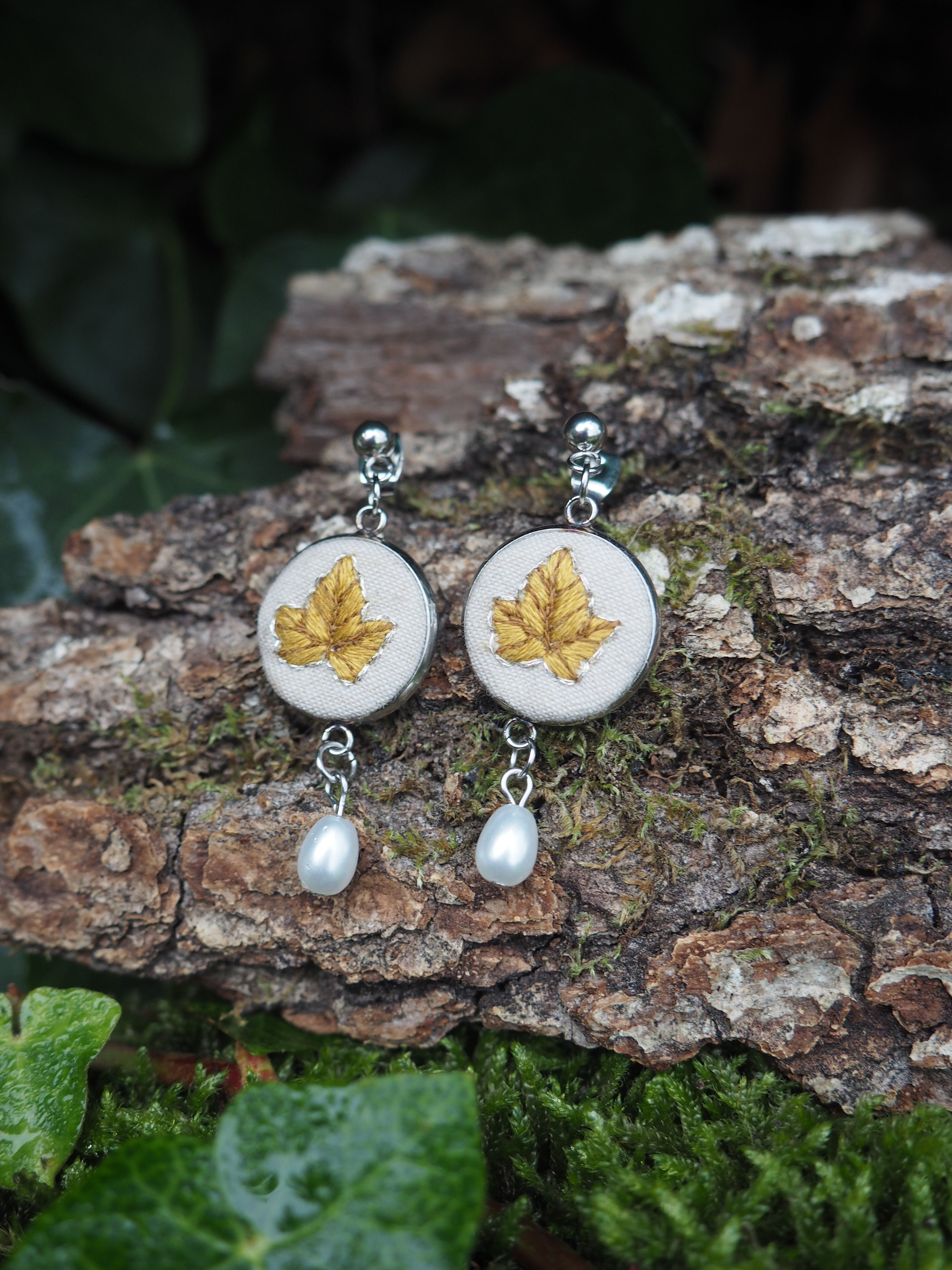 This image features a pair of elegant earrings resting on a piece of tree bark, surrounded by lush green foliage. The earrings are designed with a circular, silver-toned metal frame. Within each circle, there is a light-colored, possibly cream or off-white, fabric background on which a detailed golden-yellow leaf is embroidered. The leaf design appears to be a maple or ivy leaf, with visible veins and a slightly textured appearance from the embroidery. Suspended from the bottom of each circular earring is a single, lustrous white pearl, adding a touch of classic sophistication. The pearls are teardrop-shaped and reflect light softly. The background consists of a rough, textured piece of tree bark, covered in patches of vibrant green moss, suggesting a natural, outdoor setting. Below the bark, and slightly out of focus, are dark green leaves, possibly ivy, some of which appear to have water droplets on them, indicating a damp environment. The overall lighting is soft, highlighting the details of the earrings and the natural textures of the surroundings.