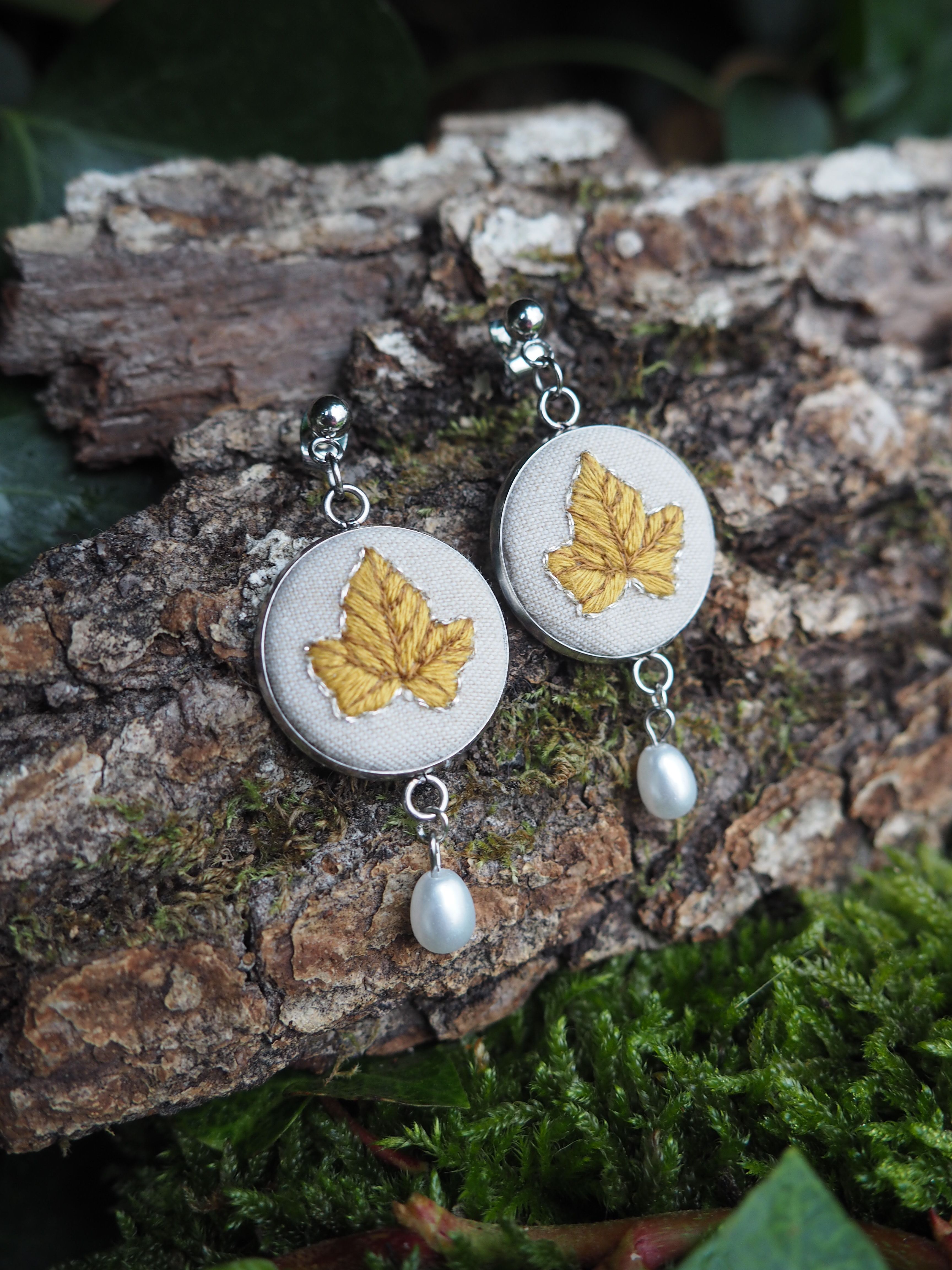 Two embroidered earrings are displayed on a piece of tree bark covered with moss. The earrings are circular, with a light beige fabric background. Each earring features a detailed embroidery of a golden-yellow leaf, possibly an ivy leaf, with visible veins. Below each circular embroidered piece, a small, lustrous white pearl dangles. The earrings are attached to silver-colored hooks. The natural setting of the bark and moss provides a rustic and earthy backdrop for the handcrafted jewelry.