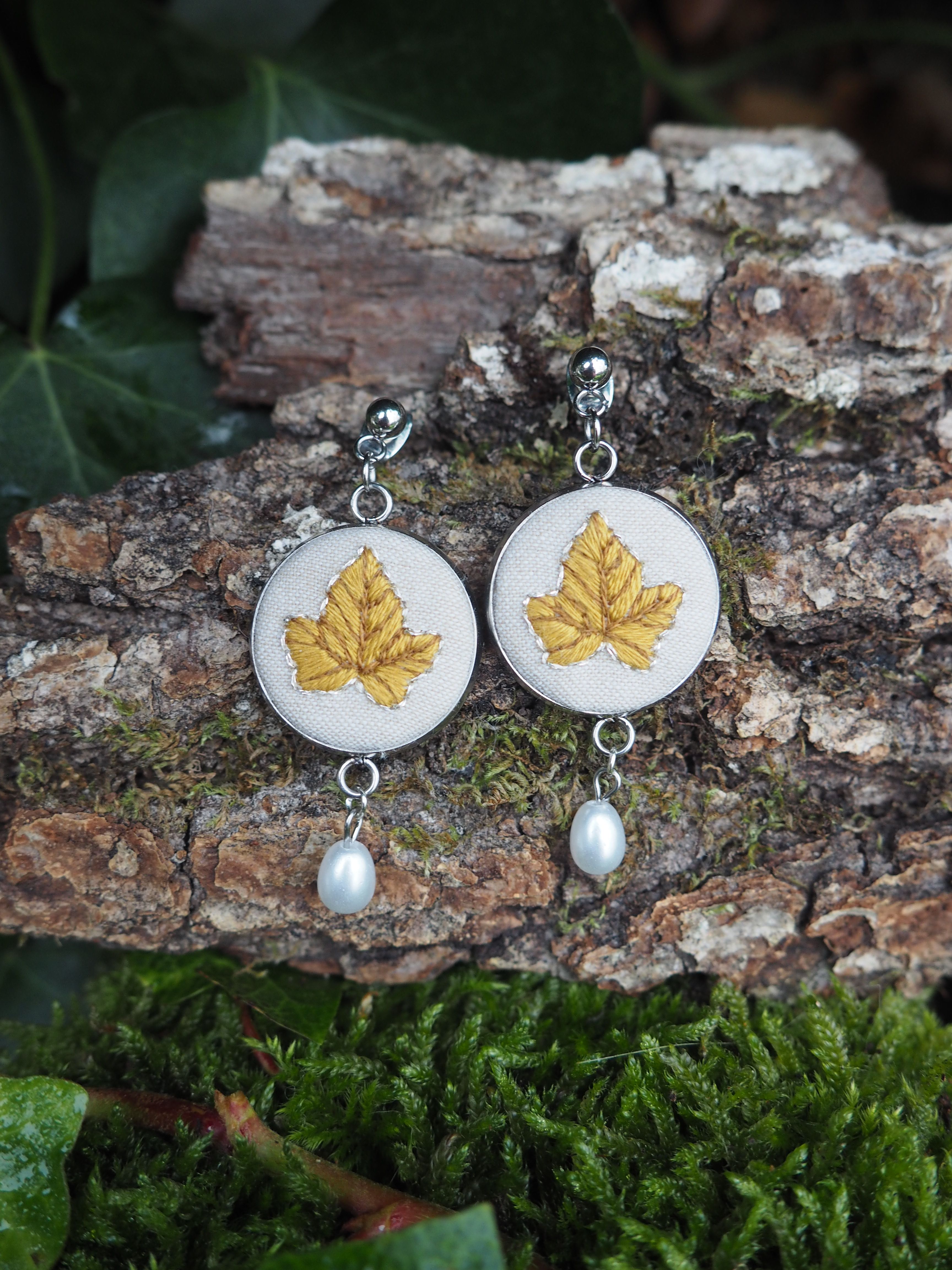 This image features a pair of elegant, nature-inspired earrings resting on a textured piece of tree bark, surrounded by lush green moss and ivy. The earrings are silver-toned and dangle, each featuring an oval-shaped pendant. The oval pendants have a light, possibly white or cream-colored fabric background, on which a detailed golden-yellow leaf is embroidered. The embroidery shows the veins and shape of the leaf clearly. Below each oval pendant, a single, lustrous white pearl hangs, adding a touch of classic sophistication. The tree bark serving as the backdrop is rich in texture, with deep grooves and lighter patches, suggesting an aged and natural surface. Patches of vibrant green moss cling to the bark, enhancing the organic feel of the scene. In the foreground and background, dark green ivy leaves are visible, further emphasizing the natural, woodland aesthetic. The lighting appears soft and natural, highlighting the details of the embroidery and the pearls, while creating a gentle contrast with the earthy tones of the bark and the vivid greens of the foliage. The overall impression is one of handcrafted beauty and natural elegance.