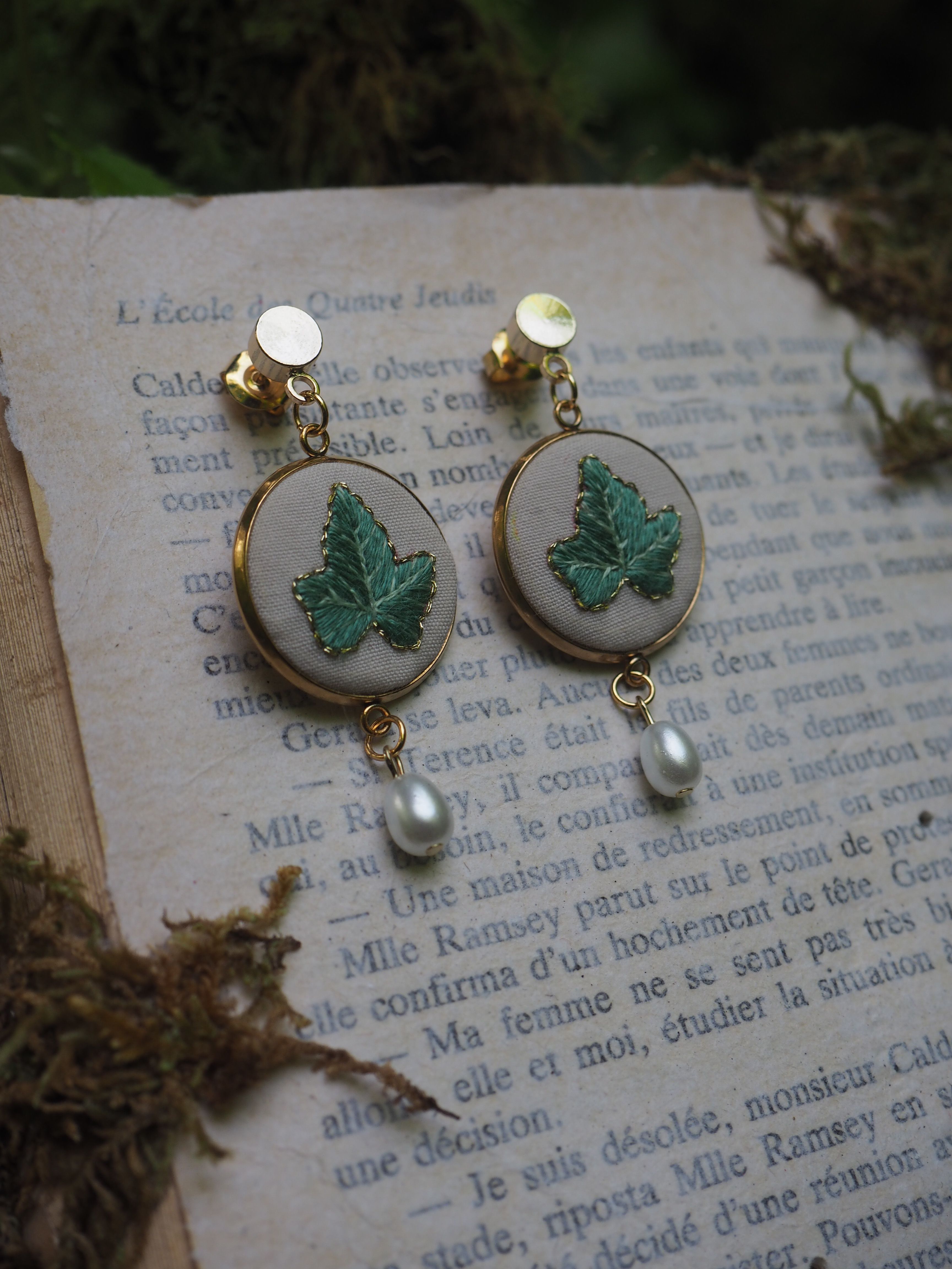 Cette image montre une paire de boucles d'oreilles élégantes posées sur une page de livre en français. Les boucles d'oreilles sont composées de plusieurs éléments : 1. Des clips ou clous dorés en haut
2. Un médaillon rond avec un fond clair (probablement beige ou blanc)
3. Une feuille verte brodée ou peinte sur chaque médaillon
4. Une petite perle blanche suspendue au bas de chaque boucle d'oreille Le style est très romantique et vintage, avec un aspect fait main ou artisanal. Les boucles d'oreilles sont photographiées sur ce qui semble être une page d'un livre ancien en français, créant une ambiance nostalgique et poétique. L'ensemble est très délicat et féminin, parfait pour un style bohème ou romantique.
