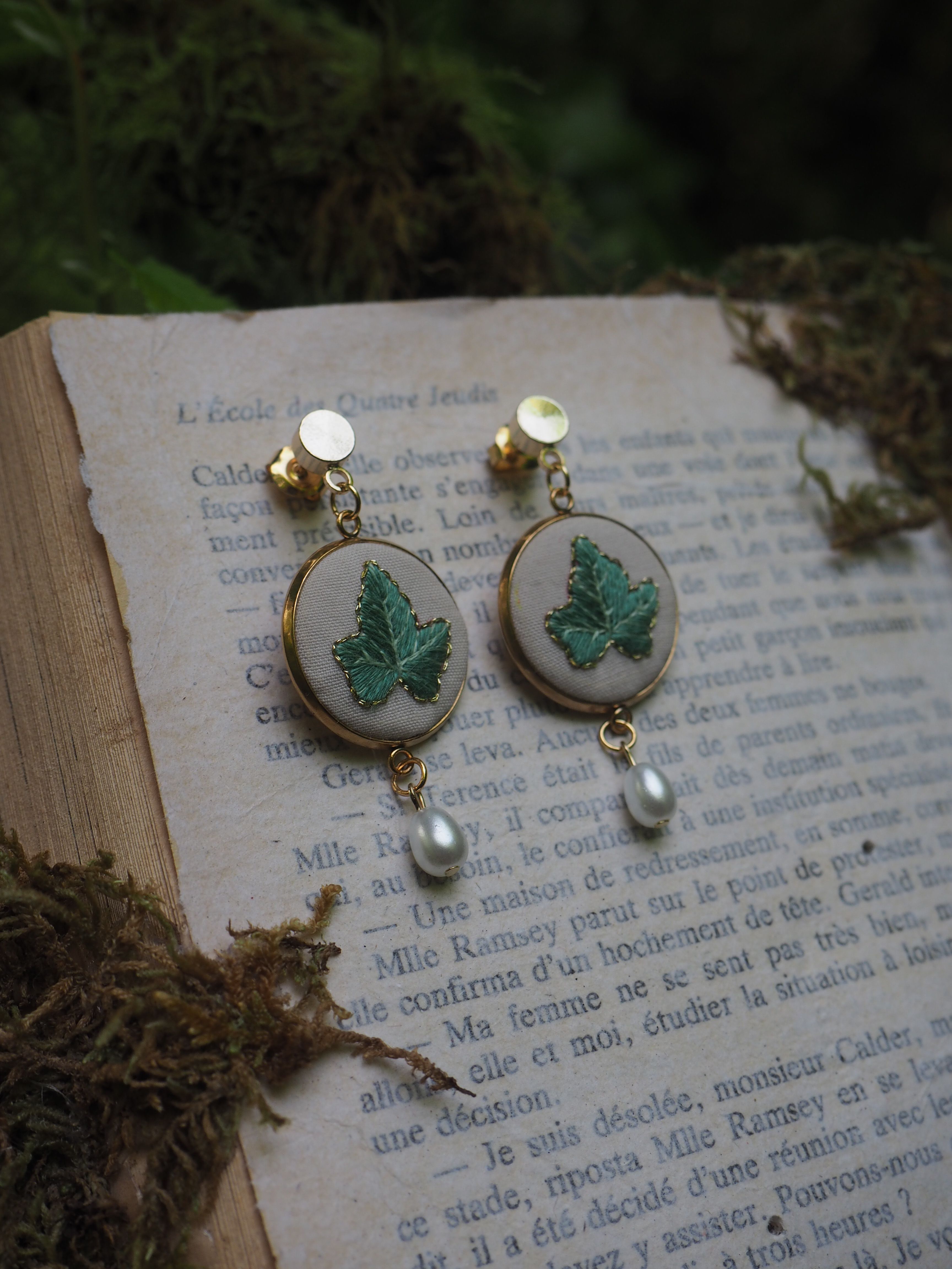 Cette image montre une paire de boucles d'oreilles élégantes posées sur une page de livre en français. Les boucles d'oreilles sont composées de médaillons ronds blancs ornés de papillons verts brodés ou peints. Au bas de chaque boucle d'oreille pend une petite perle nacrée. Les boucles d'oreilles semblent être de style vintage ou artisanal. L'arrière-plan montre du texte français imprimé sur une page, créant une ambiance romantique et littéraire. La photo a une atmosphère douce et poétique, avec une légère présence de verdure floue en arrière-plan.
