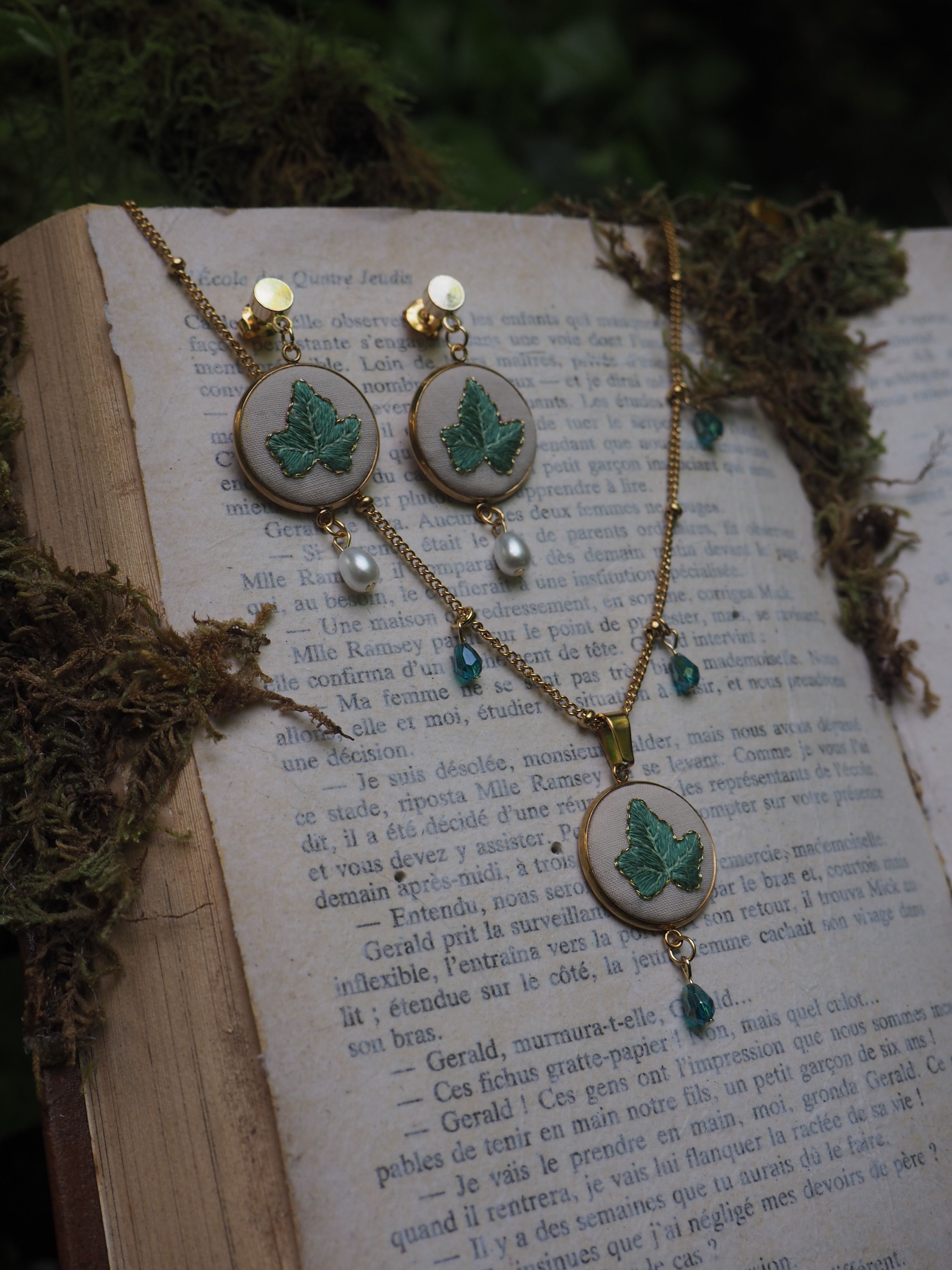 Cette image montre un ensemble de bijoux délicat posé sur un livre ouvert. L'ensemble se compose d'une paire de boucles d'oreilles et d'un collier assortis. Chaque pièce présente une broderie verte en forme de feuille ou de trèfle sur un fond blanc, encadrée dans ce qui semble être des cercles en métal doré. Les bijoux sont ornés de petites perles et de cristaux bleu-vert qui pendent en dessous des motifs brodés. L'arrière-plan montre les pages d'un livre en français avec du texte imprimé, et il y a ce qui semble être de la mousse ou des éléments naturels séchés disposés autour des bijoux, créant une ambiance naturelle et romantique. L'ensemble a une esthétique très délicate et vintage, avec une belle harmonie entre les éléments naturels et les bijoux artisanaux.