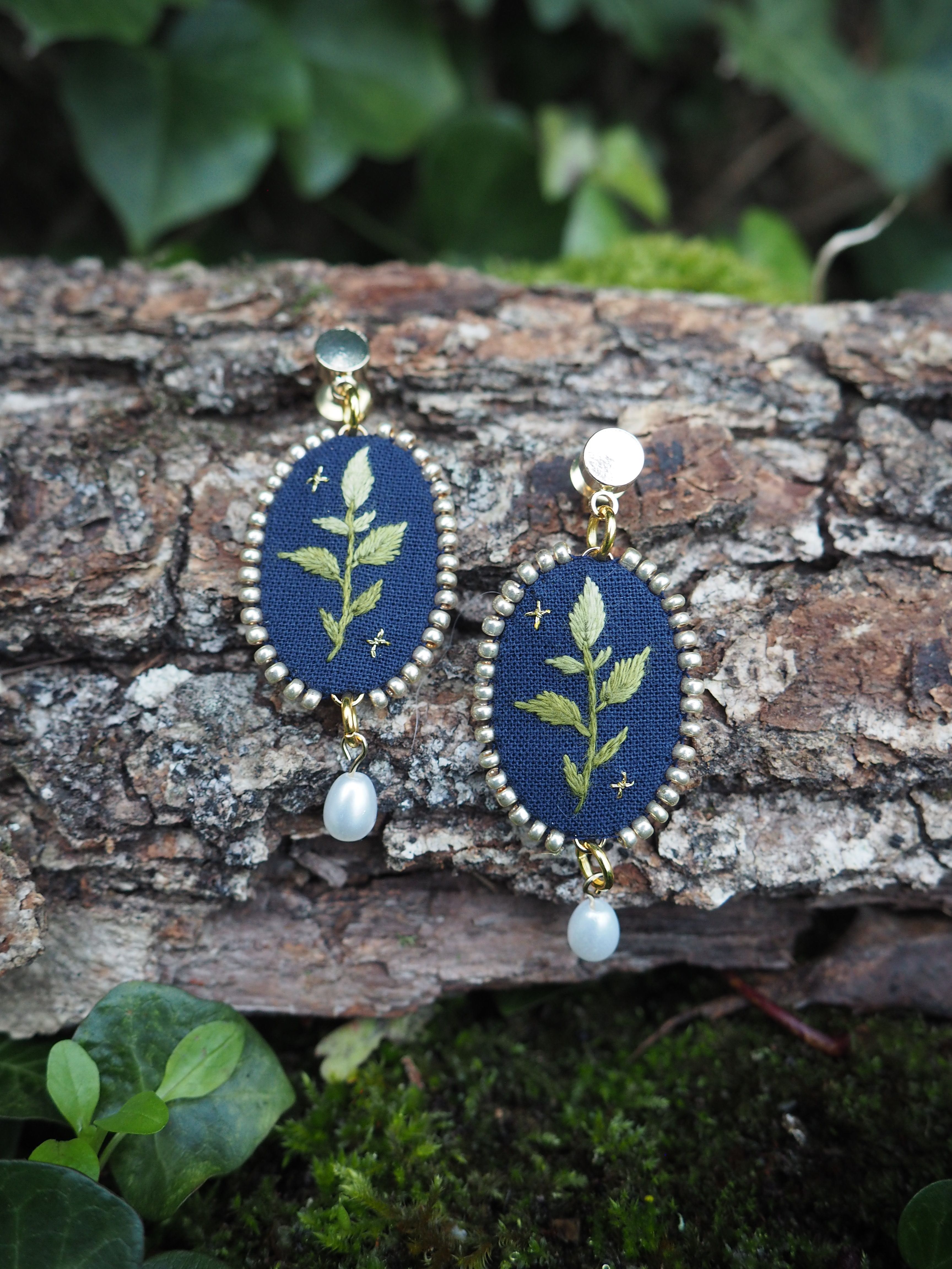 L'image montre une paire de boucles d'oreilles délicates et artisanales posées sur un morceau d'écorce d'arbre. Les boucles d'oreilles ont une forme ovale et sont composées d'un fond bleu foncé sur lequel est brodé un motif de feuille ou de branche verte. Elles sont entourées d'un cadre doré et ornées d'une petite perle blanche suspendue en bas. Le cadre naturel de l'image met en valeur ces bijoux faits main. L'écorce rugueuse et texturée contraste avec la finesse des boucles d'oreilles. En arrière-plan et autour, on peut voir de la verdure floue, probablement des feuilles, ce qui donne une ambiance naturelle et forestière à la scène. Cette photographie semble mettre en avant l'artisanat et la beauté des bijoux inspirés par la nature, en les plaçant dans un environnement qui reflète leur thème botanique.